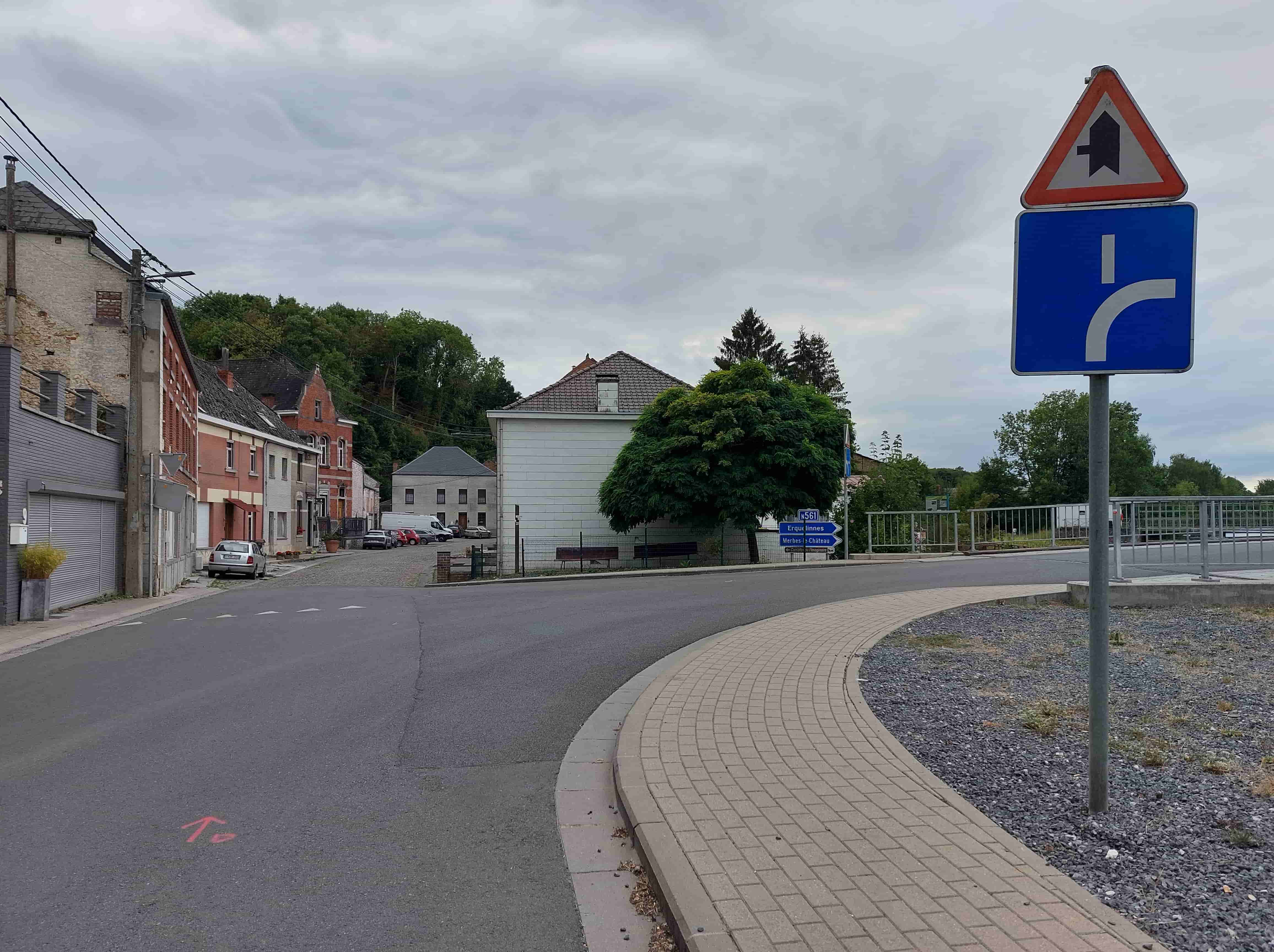 En tant qu'expert en code de la route belge, voici une description détaillée de la situation routière présentée dans l'image, en lien avec la question posée :
Description Détaillée de la Situation Routière (Examen de Conduite Belge)
L'image présente une intersection dans une zone qui semble être un village ou une petite ville, sous un ciel couvert. La perspective est celle du conducteur arrivant sur cette intersection.
Type de situation routière :
Il s'agit d'une intersection complexe où une voie principale semble rencontrer une voie secondaire et où la voie principale elle-même pourrait se diviser ou changer de direction. Plus précisément :
Une rue débouche sur la gauche.
Une voie semble continuer tout droit ou légèrement sur la droite, formant la "voie publique d'en face" mentionnée dans la question.
L'ensemble se situe dans une zone bâtie avec des maisons des deux côtés de la rue de gauche, et des arbres et de la verdure en arrière-plan, suggérant une sortie de zone urbaine ou un quartier résidentiel.
Éléments visibles :
Véhicules :
Une voiture de couleur argentée est garée sur le côté gauche de la rue latérale, face à l'arrière-plan.
Plusieurs autres véhicules (un utilitaire blanc, une voiture rouge, une voiture sombre) sont stationnés plus loin dans la rue de gauche, dans ce qui semble être un petit parking ou un élargissement de la chaussée.
Aucun véhicule en mouvement n'est visible.
Panneaux de signalisation :
À droite, sur un même poteau, deux panneaux superposés :
Panneau supérieur (triangulaire à bord rouge, fond blanc, symbole noir) : Il s'agit du panneau de danger A21b (ou A21c selon la forme précise de l'intersection), signifiant "Avertissement d'une intersection avec une route dont les conducteurs venant de gauche doivent céder le passage". Le trait principal (vertical, plus épais) représente la route sur laquelle nous nous trouvons (la route prioritaire), et le trait horizontal partant de la gauche (plus fin) représente la route qui débouche sur la nôtre. Ce panneau indique donc que les véhicules venant de la rue de gauche doivent céder le passage aux véhicules circulant sur notre voie.
Panneau inférieur (rectangulaire bleu, symbole blanc) : Il s'agit du panneau de signalisation de direction F14b, représentant une "schéma d'intersection". La ligne verticale épaisse représente la voie principale sur laquelle nous nous trouvons. La ligne courbée vers la droite indique une sortie ou un embranchement vers la droite. Cela confirme que nous sommes sur une voie principale et donne une indication de la configuration de l'intersection devant nous.
Plus loin à gauche, derrière un arbre, sur un poteau bleu :
Un panneau de direction bleu qui indique la numérotation de la route "N 561" et les directions vers "Erquelinnes", "Merbes-le-Château" et "Carnavals de l'Imaginaire". Ce panneau confirme la nature de la route sur laquelle nous nous trouvons (une route nationale) et les destinations possibles.
Feux de signalisation : Aucun feu tricolore n'est visible.
Marquages au sol :
Aucun marquage au sol de priorité (ligne discontinue de céder le passage, ligne continue de stop) n'est visible pour notre voie à l'approche de l'intersection immédiate.
Une petite marque rouge, ressemblant à une flèche ou une lettre "To", est visible sur l'asphalte en bas à gauche de l'image. Il s'agit probablement d'un marquage temporaire de chantier ou de topographie, sans pertinence pour les règles de circulation.
La chaussée est asphaltée. La rue de gauche semble débuter par des pavés avant de devenir de l'asphalte.
Autre infrastructure :
Trottoir pavé sur la droite de la voie, bordé d'un accotement en graviers.
Des bâtiments (maisons) de part et d'autre de la rue de gauche.
Un grand arbre feuillu au centre de l'image.
Une barrière métallique (garde-corps) est visible plus loin sur la droite, protégeant probablement une zone en contrebas (éventuellement un pont ou un fossé).
Poteaux électriques et lignes aériennes.
Un banc public est visible sur la droite, derrière une petite clôture.
Position et direction des véhicules (y compris le conducteur) :
Le conducteur est positionné sur une route asphaltée, clairement identifiée comme une route principale (N561) et prioritaire (par le panneau A21b). Le conducteur fait face à une intersection où une route débouche de sa gauche et où la route principale continue ou bifurque. La question indique que le conducteur "souhaite prendre la voie publique d'en face", ce qui peut signifier continuer tout droit ou tourner à gauche, traversant potentiellement le flux de circulation inverse.
Signification des panneaux de signalisation pour la situation :
Le panneau A21b est crucial : il avertit que les véhicules venant de la rue latérale de gauche doivent céder le passage à ceux qui se trouvent sur notre voie. Cela signifie que le conducteur, arrivant sur cette intersection, a la priorité sur les usagers venant de sa gauche.
Le panneau F14b confirme que nous sommes sur une voie principale et indique la configuration de la sortie/direction vers la droite.
Les panneaux de direction N561 confirment la nature de la route.
Tout autre élément pertinent pour comprendre la situation :
L'absence de tout autre panneau de priorité (tel que B1 "céder le passage" ou B5 "stop") pour notre propre voie à l'approche de l'intersection renforce l'idée que nous nous trouvons sur une route prioritaire. La question clé est de savoir si "la voie publique d'en face" implique un virage à gauche. Si c'est le cas, les règles de priorité habituelles en cas de virage à gauche s'appliqueraient, notamment céder le passage aux véhicules venant en sens inverse. Si "prendre la voie publique d'en face" signifie simplement continuer tout droit, la priorité est acquise par rapport à la voie de gauche.