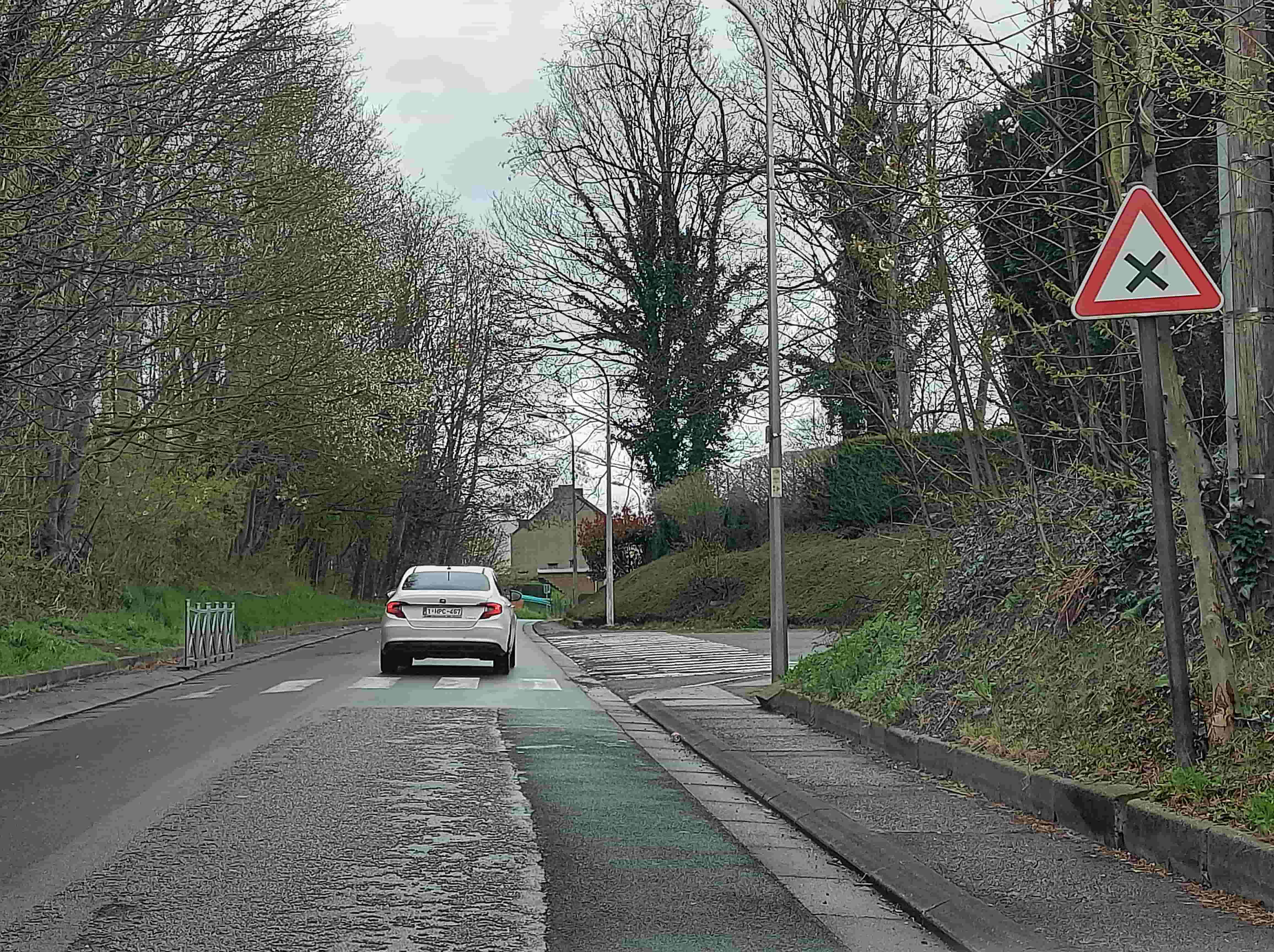 En tant qu'expert du code de la route belge, voici la description détaillée de la situation routière représentée sur l'image, en tenant compte du contexte de la question :
Description Détaillée de la Situation Routière (Examen de Conduite Belge)
L'image présente une vue de face d'une route en milieu semi-urbain ou périurbain, sous un ciel couvert, suggérant une journée nuageuse. La perspective est celle d'un conducteur qui s'apprête à poursuivre sa route en ligne droite.
Le type de situation routière :
Il s'agit d'une approche d'une intersection, signalée par un panneau spécifique.
La route comporte également un passage pour piétons (zebra crossing) et un ralentisseur (dos d'âne) intégré à ce passage.
Une bande cyclable est clairement visible sur le côté droit de la chaussée.
Les éléments visibles :
Chaussée : La surface de la route est en asphalte. Sur la droite, une bande cyclable est peinte en vert clair, séparée du reste de la chaussée. Juste avant l'intersection, la chaussée présente un ralentisseur (bosses transversales) qui s'étend sur toute la largeur de la voie de circulation et de la bande cyclable.
Marquages au sol : Un passage pour piétons est matérialisé par des bandes blanches parallèles (zebra crossing) traversant la chaussée. Une flèche blanche, à peine visible à droite du ralentisseur, pourrait indiquer une direction ou une voie, mais elle est trop effacée pour être déterminante.
Accotements et environnement : Des deux côtés de la route, des accotements verdoyants bordent la chaussée. De nombreux arbres dénudés (suggérant la fin de l'hiver ou le début du printemps) encadrent la route, surtout sur la gauche. Sur la droite, un talus herbeux monte vers un arrière-plan où l'on distingue une habitation.
Mobilier urbain : Un lampadaire est visible sur la droite. Un poteau électrique ou de télécommunication se trouve également sur la droite, supportant un petit panneau jaune rectangulaire illisible en raison de la distance et de la faible résolution. Sur la gauche, un petit muret ou une glissière métallique basse est implantée le long de l'accotement.
La position et direction des véhicules s'il y en a :
Véhicule de l'observateur : Le conducteur (vous, dans le cadre de la question) est situé sur la chaussée principale, en amont du ralentisseur et du passage pour piétons, et donc de l'intersection.
Voiture blanche : Une voiture de couleur claire (probablement blanche ou argentée, ressemblant à une Fiat Tipo) circule devant le véhicule de l'observateur, dans la même direction. Elle est positionnée juste après le passage pour piétons, potentiellement en train de le franchir ou de s'apprêter à tourner/continuer. Sa plaque d'immatriculation est visible : "1-IPC-467".
Motard (non visible sur l'image) : La question mentionne "Un motard vient de ma droite en poussant une motocyclette." Cet usager n'est pas visible sur l'image, mais il est un élément clé du scénario.
Les panneaux de signalisation présents et leur signification :
Panneau A21 : Sur la droite de la route, à hauteur de l'intersection et avant le passage pour piétons, est implanté un panneau de signalisation triangulaire à bord rouge et fond blanc, comportant une croix de Saint-André noire (deux lignes diagonales noires se croisant). Il s'agit du panneau A21, qui signifie en Belgique : "Attention, intersection où la priorité de droite est applicable." Cela indique que le conducteur doit céder le passage à tout véhicule venant de sa droite.
Autres panneaux : Un petit panneau jaune rectangulaire est visible plus loin sur la droite, sur un poteau, mais il est trop flou pour être identifié ou interprété.
Tout autre élément pertinent pour comprendre la situation :
Le contexte du "motard poussant une motocyclette" : C'est l'information la plus cruciale pour la question. En Belgique, une personne poussant une motocyclette est considérée comme un piéton par le Code de la Route. Cette classification est fondamentale pour déterminer les règles de priorité, car les règles applicables à un piéton (notamment au niveau d'un passage pour piétons ou s'il s'engage sur la chaussée) sont différentes de celles applicables à un véhicule en mouvement.
Présence du passage pour piétons et du ralentisseur : Ces éléments soulignent que la zone est une zone à risques, nécessitant une vigilance accrue et une réduction de la vitesse, notamment envers les usagers faibles. Le ralentisseur est là pour forcer les véhicules à ralentir avant et sur le passage pour piétons.
• Bande cyclable : La présence de la bande cyclable verte indique que des cyclistes sont également susceptibles d'emprunter cette voie, traversant potentiellement l'intersection ou le passage pour piétons.
En résumé, la situation se déroule à l'approche d'une intersection avec priorité de droite, où sont également présents un passage pour piétons et un ralentisseur. La question introduit un élément non visible mais crucial : un "motard" qui, en poussant sa moto, doit être considéré comme un piéton selon le code de la route belge, venant de la droite du véhicule de l'observateur.