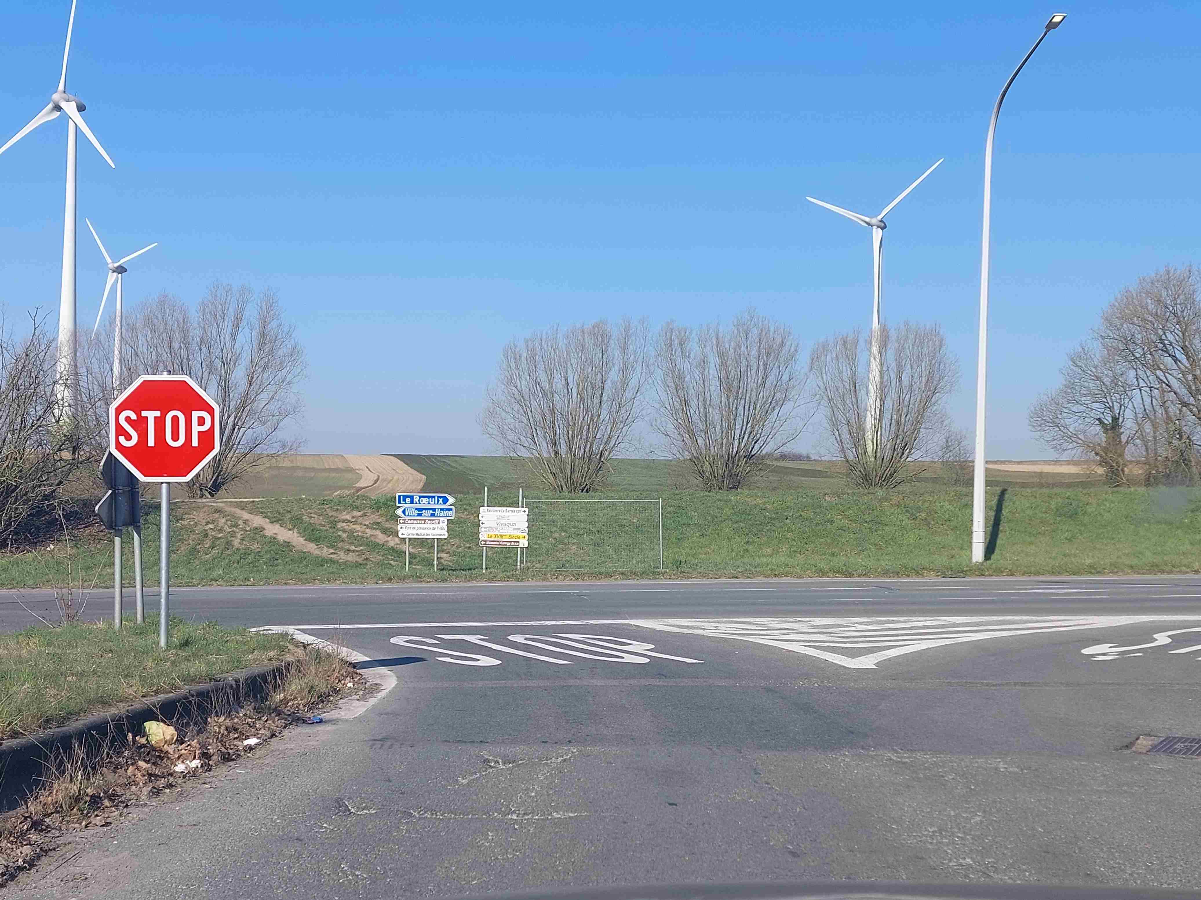 En tant qu'expert en code de la route belge, voici une analyse détaillée de l'image pour répondre à la question posée : "À ce carrefour, où dois-je marquer l'arrêt et céder le passage ?"
Description Détaillée de la Situation Routière
Type de situation routière :
L'image représente l'approche d'un carrefour en T (ou intersection en "Y" très ouvert) en milieu semi-rural ou agricole. Le conducteur (de la voiture d'où est prise la photo) se trouve sur une voie d'accès secondaire qui débouche sur une chaussée principale, qui semble plus large et potentiellement plus fréquentée. Le temps est clair et ensoleillé.
Éléments visibles :
Voie du conducteur : Une chaussée asphaltée unique, bordée par une bordure sur le côté gauche. La végétation (herbe, quelques buissons) est présente sur les accotements.
Chaussée transversale : La route que le conducteur va traverser ou sur laquelle il va s'engager. Elle est visible plus loin, au-delà de la ligne d'arrêt.
Panneaux de signalisation :
Sur le côté gauche de la voie du conducteur, se dresse un panneau de signalisation B5 "STOP" octogonal rouge avec l'inscription "STOP" en lettres blanches, cerclé de blanc. Il est fixé sur un poteau.
Au-delà de l'intersection, sur le côté gauche de la chaussée transversale, se trouvent des panneaux de direction : un panneau bleu avec des flèches blanches indiquant "Le Roeulx", "Ville-sur-Haine", "Complexe Sportif", etc., et un panneau blanc à droite donnant d'autres indications locales (Vivaqua, Le XVIII Siècle, etc.).
Marquages au sol :
Un large marquage au sol avec l'inscription "STOP" est peint en blanc sur la chaussée du conducteur, précédant une ligne.
Juste après ce marquage "STOP", une ligne d'arrêt continue et épaisse est tracée en blanc, perpendiculairement à la voie du conducteur. Cette ligne s'étend sur toute la largeur de la voie.
Végétation et paysage : Des arbres à feuilles caduques (dénudés, suggérant la fin de l'hiver ou le début du printemps) bordent les champs qui s'étendent au loin. Des champs cultivés sont visibles en arrière-plan.
Infrastructures : Plusieurs éoliennes sont visibles à l'horizon, contribuant au caractère rural. Un lampadaire moderne se dresse sur le côté droit de la chaussée transversale.
Véhicules : Aucun autre véhicule n'est visible dans l'image, ni sur la voie du conducteur, ni sur la chaussée transversale.
Position et direction des véhicules :
Le conducteur est positionné sur la voie d'accès secondaire, s'approchant de l'intersection. Son véhicule est à l'arrêt ou en phase d'approche de la ligne d'arrêt. Il fait face à l'intersection.
Panneaux de signalisation présents et leur signification (Code de la Route Belge) :
Panneau B5 "STOP" (octogonal rouge avec l'inscription "STOP" en blanc) : Ce panneau est un signal de priorité. Sa présence indique une obligation formelle pour le conducteur de marquer un arrêt complet et obligatoire à la hauteur de la ligne d'arrêt (la ligne blanche continue et épaisse tracée sur la chaussée). Après avoir marqué cet arrêt, le conducteur doit impérativement céder le passage à tous les usagers de la route circulant sur la chaussée traversée, quelle que soit leur direction (à gauche ou à droite). L'arrêt doit être total, même si la voie semble libre.
Marquage au sol "STOP" et ligne d'arrêt continue : Ces marquages confirment et renforcent l'obligation d'arrêt et indiquent précisément l'endroit où le véhicule doit s'immobiliser. La ligne d'arrêt prime sur le marquage "STOP" au sol si les deux ne sont pas parfaitement alignés, mais ici, le marquage est bien positionné avant la ligne.
Panneaux de direction (bleus) et d'information (blancs) : Ces panneaux fournissent des informations sur les destinations et points d'intérêt. Ils n'ont aucune incidence sur les règles de priorité à l'intersection pour le conducteur du véhicule d'où est prise la photo.
Tout autre élément pertinent :
L'absence de feux de signalisation, d'agents qualifiés ou d'autres panneaux de priorité (comme "cédez le passage" B1 ou B3) confirme que le panneau B5 "STOP" est le seul et unique régulateur de priorité pour le conducteur arrivant à cette intersection. L'emplacement de la ligne d'arrêt et du panneau "STOP" indique clairement l'obligation d'arrêt avant de s'engager sur la chaussée principale. La visibilité vers la chaussée transversale semble dégagée, ce qui est essentiel pour évaluer le trafic après l'arrêt obligatoire.