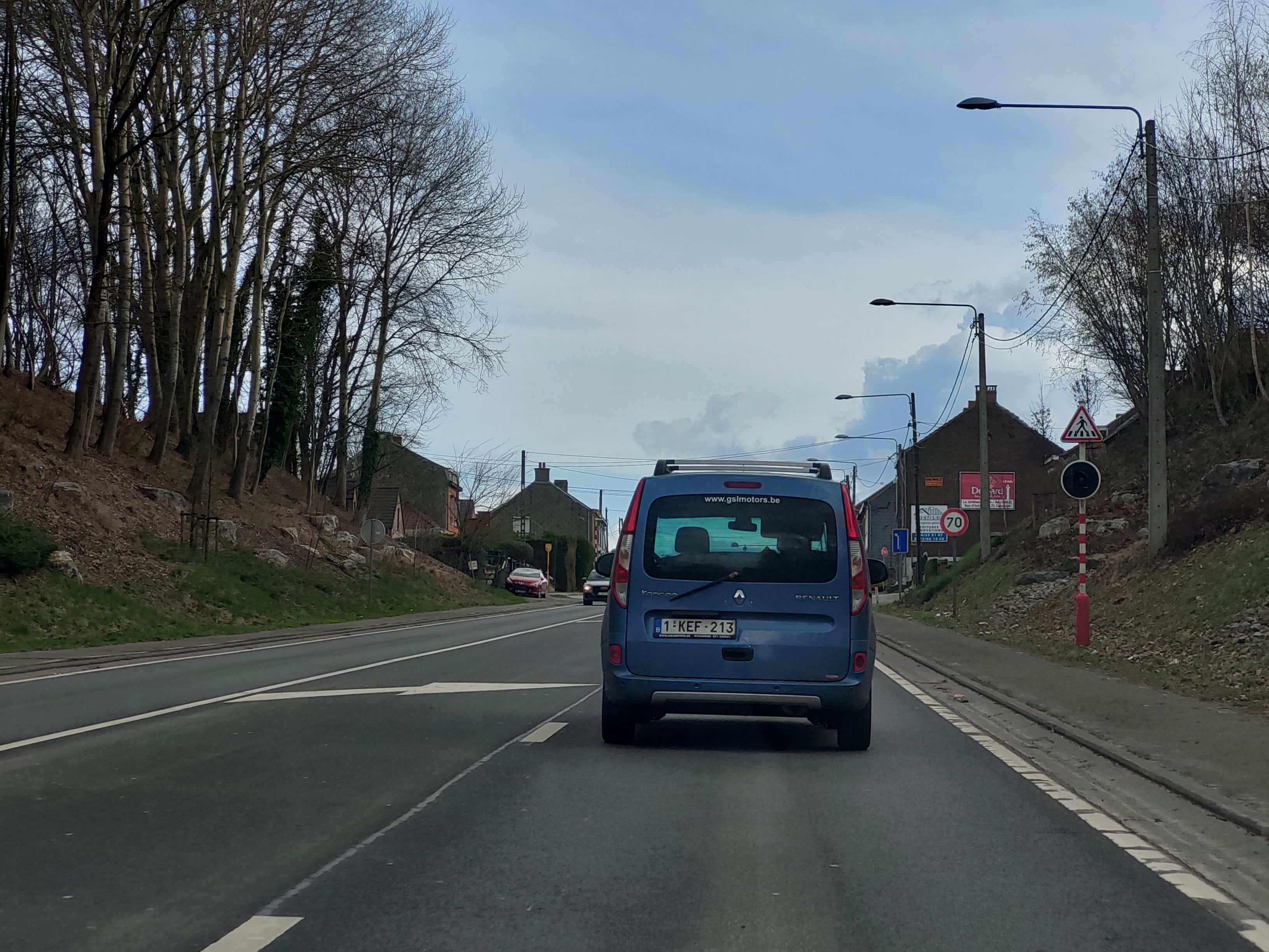 En tant qu'expert en code de la route belge, voici une analyse détaillée de l'image, mettant l'accent sur le contexte de la question relative aux "flèches blanches de rabattement".
Description détaillée de la situation routière:
L'image présente une vue depuis l'intérieur d'un véhicule circulant sur une route bidirectionnelle (ou potentiellement une route à chaussée divisée/plusieurs voies dans le même sens qui se réduisent) en agglomération ou à l'approche de celle-ci, comme l'indiquent les habitations et les panneaux de signalisation. Le temps est couvert, et les arbres sont défeuillés, suggérant l'hiver ou le début du printemps.
Type de situation routière:
Il s'agit d'une section de route où le nombre de voies disponibles dans le sens de circulation du véhicule photographiant est sur le point de diminuer. La présence de marquages au sol spécifiques et de panneaux de signalisation indique une zone nécessitant une vigilance accrue, notamment à l'approche d'un passage pour piétons et d'une limitation de vitesse. La route semble légèrement en pente montante ou en courbe douce vers la gauche.
Éléments visibles:
Véhicules:
Un monospace bleu de marque Renault (probablement un Kangoo) avec une plaque d'immatriculation belge "1-KEF-213" est directement devant le véhicule du conducteur. Il est équipé de barres de toit et arbore un autocollant "www.gslmotors.be" sur la lunette arrière. Il circule dans la voie de droite.
Plus loin, dans le sens inverse de circulation, un véhicule de couleur rouge est visible, et derrière lui, un véhicule plus sombre (ressemblant à une Audi).
Marquages au sol:
Dans la voie de droite, là où le véhicule bleu circule, deux flèches blanches de rabattement sont clairement visibles. Ces flèches, de forme allongée et pointant vers la gauche, indiquent aux conducteurs que la voie qu'ils occupent va prendre fin (se rabattre) et qu'ils doivent se déporter vers la voie adjacente de gauche.
Une ligne continue blanche marque la limite droite de la chaussée (accotement ou bas-côté).
Une ligne discontinue blanche sépare la voie de droite de la voie de gauche dans le même sens de circulation, permettant le rabattement.
Plus loin, on aperçoit ce qui semble être une ligne continue centrale séparant les deux sens de circulation.
Panneaux de signalisation (visibles sur la droite de la route, dans le sens de la marche):
Un panneau de danger A21 (triangle blanc bordé de rouge, avec un pictogramme noir de deux piétons) signalant une traversée de piétons ou un endroit fréquenté par des piétons. Il incite à la prudence et à modérer sa vitesse.
Sous ce panneau A21, un signal lumineux rond et noir, éteint (vraisemblablement un feu pour piétons), est monté sur un poteau rayé rouge et blanc, souvent utilisé pour les passages à niveau ou les obstacles.
Plus loin et un peu en retrait, un panneau de limitation de vitesse C43, indiquant "70" (rond blanc bordé de rouge, avec le chiffre 70 noir), signifie que la vitesse maximale autorisée est de 70 km/h. Ce panneau est placé à côté de panneaux publicitaires ("DEWEZ", "VOITURES D'OCCASION").
Un panneau de direction D1b (carré bleu avec une flèche blanche pointant vers la gauche) est également visible, juste avant le panneau "70". Ce panneau indique une direction obligatoire pour une voie ou une sortie spécifique, ou une direction générale.
Autres éléments:
Des maisons résidentielles sont visibles de part et d'autre de la route, certaines semblant anciennes.
Des arbres, pour la plupart sans feuilles, bordent la route, notamment sur les talus en pente.
Plusieurs lampadaires d'éclairage public sont alignés le long de la route.
Des lignes électriques ou téléphoniques aériennes sont visibles.
Position et direction des véhicules:
Le véhicule de l'observateur circule tout droit, derrière le monospace bleu Renault. Le monospace bleu est positionné dans la voie de droite, au niveau des premières flèches de rabattement, et s'apprête donc à se déporter sur la voie de gauche. Les autres véhicules visibles circulent dans le sens inverse.
Signification des panneaux:
A21 (Triangle, piétons): Danger, traversée de piétons ou endroit fréquenté par des piétons. Réduire sa vitesse et être prêt à s'arrêter.
C43 (Rond, 70): Interdiction de dépasser la vitesse de 70 km/h.
• D1b (Carré bleu, flèche gauche): Indique une direction obligatoire, généralement pour une voie, une sortie ou un itinéraire spécifique.
Contexte de la question "Ces flèches blanches de rabattement :":
Les flèches blanches de rabattement peintes sur la chaussée signalent la suppression prochaine de la voie de circulation sur laquelle elles sont dessinées. Elles imposent aux conducteurs de cette voie de se rabattre vers la ou les voies situées à leur gauche. Elles sont un avertissement précoce pour permettre aux conducteurs d'anticiper le changement de voie en toute sécurité. Dans cette situation, le conducteur du véhicule bleu, et par extension celui qui le suit, doit se préparer à changer de voie vers la gauche. La présence simultanée du panneau "piétons" et de la limitation à 70 km/h renforce l'idée d'une zone où la prudence est de mise.