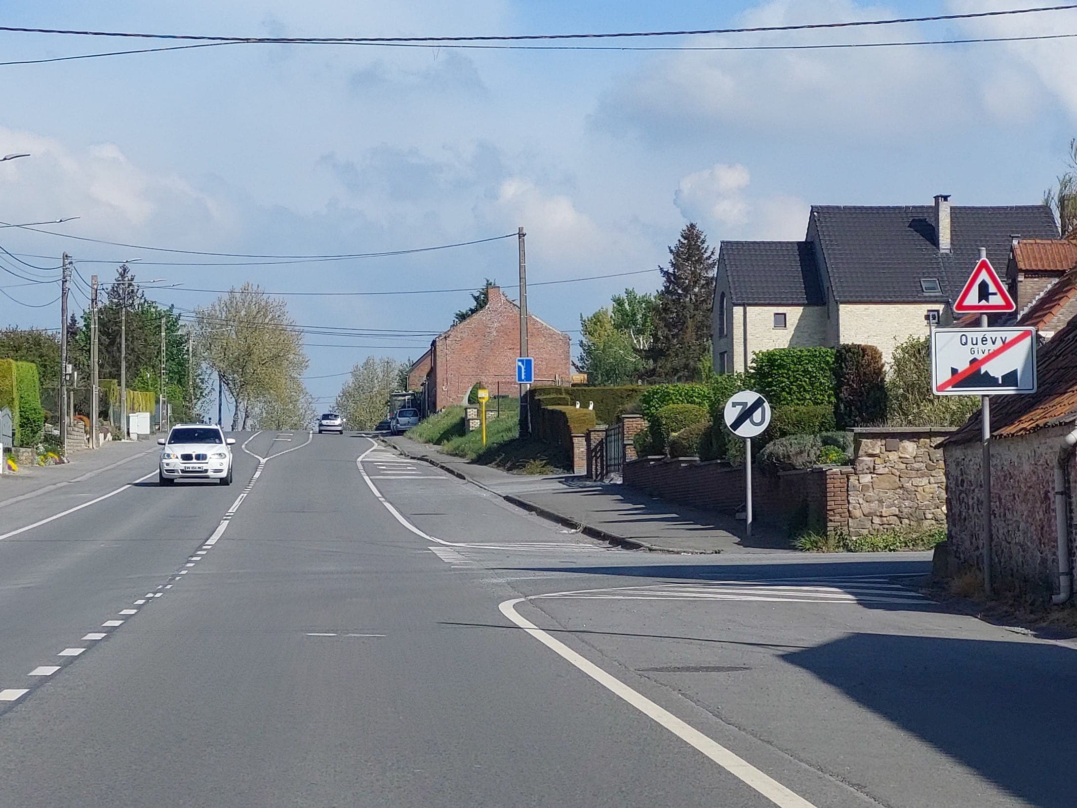 En tant qu'expert en code de la route belge, voici une description détaillée de la situation routière présentée sur l'image, en se concentrant sur les éléments pertinents pour la question posée.
Description détaillée de la situation routière (Examen de conduite - Wallonie)
Type de situation routière :
L'image présente une portion de route bidirectionnelle (une voie par sens de circulation) en dehors d'une agglomération ou à sa limite. Il s'agit d'une route asphaltée, avec un accotement aménagé sur la droite (trottoir ou bas-côté stabilisé). Sur la droite, une intersection est clairement visible, où une route secondaire débouche. La situation suggère une zone semi-rurale ou de transition entre le bâti et la campagne.
Les éléments visibles :
La route : Large et en bon état, elle est marquée par une ligne blanche discontinue centrale séparant les deux sens de circulation. Sur la gauche, la ligne discontinue s'élargit en une zone hachurée, pouvant indiquer une zone d'attente pour tourner à gauche ou un aménagement routier spécifique. Une ligne blanche continue délimite le bord droit de la chaussée. Un passage pour piétons (bandes blanches transversales) est visible sur la droite, juste avant le groupe de panneaux.
Véhicules :
Une voiture blanche de type SUV (BMW X5, immatriculation belge 1-VVW-654) approche en sens inverse, au centre de sa voie.
Une autre voiture claire est visible au loin, également en sens inverse.
Une voiture de couleur grise est stationnée sur le côté gauche de la route, plus loin.
Panneaux de signalisation (à droite, dans le sens de la marche) :
La question fait référence à "3 signaux qui sont situés à ma droite". En partant du plus proche vers le plus éloigné (mais toujours dans le groupe principal à droite) :
Panneau C43 "Fin d'agglomération" : Le grand panneau rectangulaire blanc, bordé de noir, avec le nom "Quévy" (et "Givry" en dessous) barré d'une ligne rouge diagonale. Une silhouette de ville barrée est également présente. Ce panneau indique que le conducteur quitte l'agglomération de "Quévy".
Panneau A21 "Carrefour où la priorité est à droite" : Le panneau triangulaire à bord rouge, avec une croix noire symbolisant un carrefour où la priorité de droite est d'application. Il est situé juste au-dessus du panneau C43. Ce panneau signale la présence d'un carrefour où les véhicules venant de droite (de la petite route débouchant sur la droite) sont prioritaires.
Panneau C47 "Fin de la limitation de vitesse à 70 km/h" : Le panneau circulaire blanc, bordé de noir, avec le chiffre "70" barré d'une ligne noire diagonale. Ce panneau annule une précédente limitation de vitesse à 70 km/h. Il est situé légèrement en retrait et à gauche du panneau C43/A21.
Autre panneau visible mais moins pertinent pour la vitesse au-delà des 3 signaux clés : Plus loin sur la droite, un panneau carré bleu avec une flèche blanche pointant vers la droite (panneau D3 "Direction obligatoire à droite" ou "Indication de direction à suivre") est visible à hauteur du carrefour, partiellement masqué par la végétation. Il indique une direction à prendre ou à suivre, mais n'est pas un signal direct de vitesse ou un des 3 signaux principaux pour cette question.
Feux : Aucun feu de signalisation n'est visible.
Marquages au sol : Outre les lignes de circulation, un passage pour piétons est présent juste avant le groupe de panneaux de droite. Des lignes discontinues blanches sont visibles au niveau de l'intersection de droite, indiquant un cédez-le-passage implicite pour la voie débouchant de droite (mais le panneau A21 prime pour la priorité).
Infrastructure et paysage :
Des habitations (maisons en briques ou en pierre) sont visibles des deux côtés de la route, plus denses sur la droite.
Des haies et des murs de pierre ou de briques délimitent les propriétés.
Des poteaux électriques avec des lignes aériennes longent la route.
De la végétation (arbres, buissons) est présente en abondance.
Le ciel est dégagé avec quelques nuages, indiquant un temps ensoleillé.
Position et direction des véhicules s'il y en a :
Le conducteur qui pose la question se trouve sur la route principale et se dirige vers les panneaux de signalisation à droite (donc il s'éloigne de l'appareil photo). La voiture blanche SUV vient en sens inverse. La voiture lointaine vient aussi en sens inverse. La voiture garée est sur l'accotement gauche, dans le sens de la marche de la voiture blanche.
Panneaux de signalisation présents et leur signification (détail pour les 3 signaux clés) :
Les trois signaux situés à droite et pertinents pour la question de la vitesse maximale sont :
Le panneau C43 "Fin d'agglomération Quévy Givry" : Signale que le conducteur quitte l'agglomération de Quévy (et se dirige vers Givry). En Belgique, la vitesse maximale autorisée en agglomération est de 50 km/h. La fin d'agglomération signifie un retour à la réglementation générale des routes hors agglomération, sauf indication contraire.
Le panneau A21 "Carrefour où la priorité est à droite" : Avertit de la présence d'un carrefour où la règle de la priorité de droite doit être appliquée. Bien qu'il ne s'agisse pas directement d'un panneau de limitation de vitesse, il indique une situation où la prudence est requise et où la vitesse doit être adaptée.
Le panneau C47 "Fin de la limitation de vitesse à 70 km/h" : Annule une restriction de vitesse précédente de 70 km/h. Cela signifie que la limitation de 70 km/h qui était en vigueur prend fin à partir de ce point.
Tout autre élément pertinent :
Le contexte est la Wallonie. Il est crucial de considérer l'effet combiné de ces trois panneaux sur la vitesse. Le fait de quitter une agglomération (C43) et la fin d'une limitation spécifique à 70 km/h (C47) sont les éléments déterminants pour établir la vitesse maximale autorisée au-delà de ces signaux. La priorité de droite (A21) est une règle de comportement à l'intersection, mais n'établit pas une limite de vitesse chiffrée.