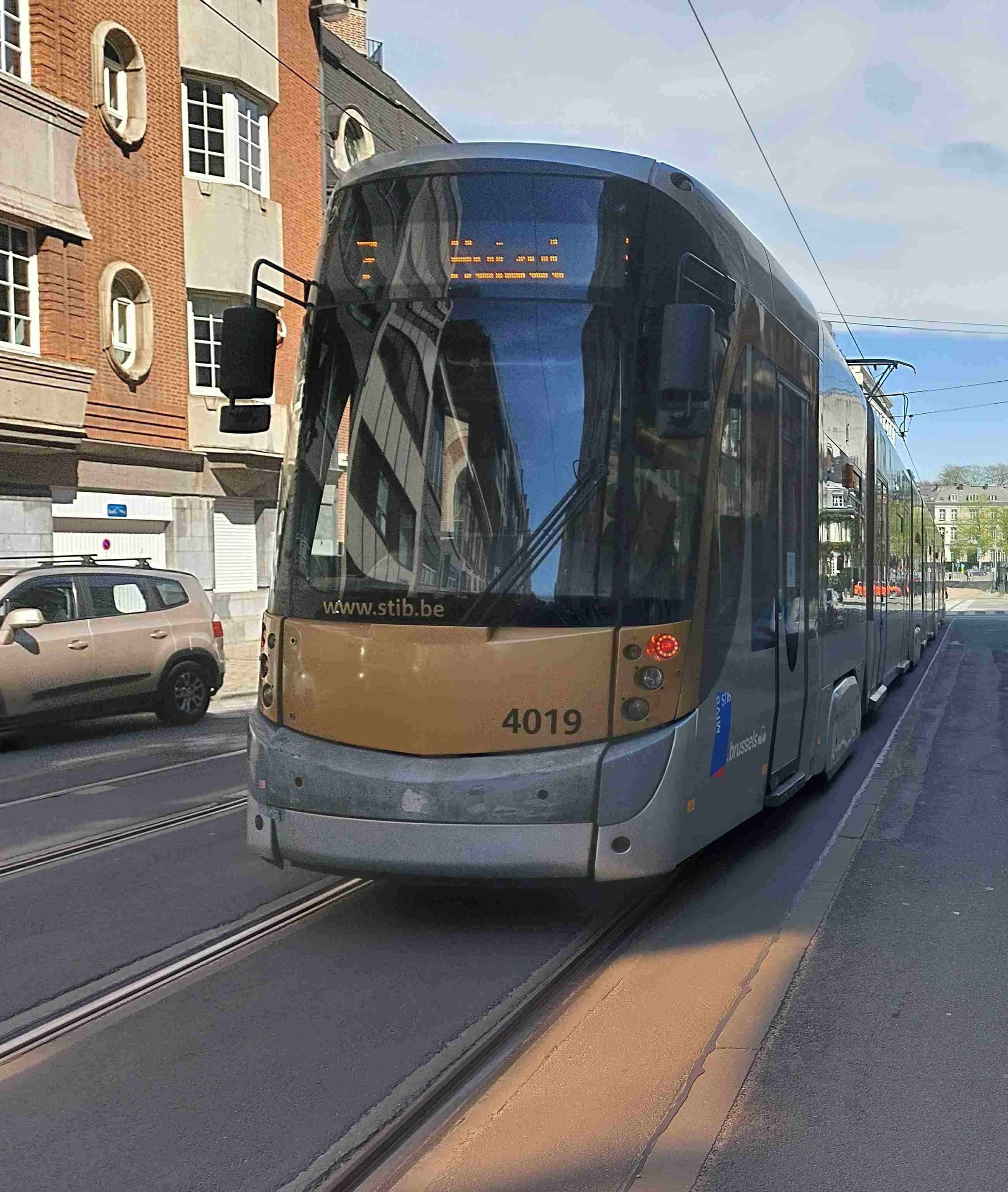 En tant qu'expert en code de la route belge, voici une description détaillée de la situation routière présentée dans l'image, en tenant compte du contexte fourni : "Le tram est immobilisé pour faire descendre et monter des passagers, les portes s'ouvrent."
Description Détaillée de la Situation Routière (Belgique)
L'image présente une scène de rue urbaine typique de Bruxelles, caractérisée par la présence d'un tram en stationnement et d'autres éléments de l'infrastructure routière et urbaine.
Type de situation routière:
Il s'agit d'une voie de circulation urbaine, probablement une rue à sens unique ou à double sens avec des voies de tram intégrées dans la chaussée. Le cadre est résidentiel ou commercial, comme en témoignent les bâtiments le long de la rue.
Le tram est actuellement immobilisé en station, comme l'indique le contexte de la question ("faire descendre et monter des passagers, les portes s'ouvrent"). Cela implique un arrêt à un point d'arrêt de tramway, potentiellement sans quai dédié, où les passagers monteraient ou descendraient directement sur la chaussée ou un accotement bas.
Il ne s'agit pas d'une intersection clairement visible, mais d'un tronçon de route rectiligne où la circulation est partagée avec le tram.
Éléments visibles:
Véhicules:
Tram: Un tramway moderne de la STIB (Société des Transports Intercommunaux de Bruxelles), de couleur gris et doré, avec le numéro d'exploitation "4019" affiché à l'avant. L'inscription "www.stib.be" et "Brussels" est également visible. Le tram est orienté vers le spectateur, mais légèrement en biais, suggérant qu'il avance ou s'est arrêté en direction de la perspective. Les feux d'avertissement rouges (ou feux arrière si la perspective était légèrement différente, mais l'emplacement suggère des feux de position/urgence) sont allumés sur le côté droit de l'avant du tram (depuis la perspective du tram). La destination affichée à l'avant (sur l'écran LED) est visible en caractères orange, mais illisible. Le pantographe (bras articulé pour la prise de courant) est visible sur le toit du tram, raccordé aux lignes aériennes.
Voiture: Une voiture de type SUV/crossover de couleur beige/dorée est visible sur le côté gauche de l'image, partiellement dissimulée par le tram et les bâtiments. Elle semble être garée ou arrêtée le long du trottoir, dans la même direction que celle dans laquelle le tram est supposé circuler. Elle est équipée de barres de toit.
Panneaux de signalisation:
Un panneau E1 "Stationnement Interdit" (rond avec fond bleu, cercle rouge et barre diagonale rouge) est visible sur la façade du bâtiment à gauche, juste au-dessus de la voiture beige. Ce panneau interdit le stationnement sur la bande de roulement où il est placé.
Un autre panneau rectangulaire bleu avec un pictogramme blanc (difficilement identifiable en raison de la distance et de la faible résolution, potentiellement une information sur un sens unique, une impasse, ou une obligation directionnelle) est visible au-dessus du panneau E1.
Feux de signalisation:
Aucun feu de signalisation de carrefour n'est visible.
Les feux rouges du tram (mentionnés précédemment) sont allumés.
Marquages au sol:
Des rails de tramway sont clairement incrustés dans la chaussée, délimitant la voie du tram.
Aucun autre marquage au sol spécifique (lignes de dépassement, zébras, passages pour piétons, etc.) n'est visible dans le champ de vision immédiat devant le tram ou à ses côtés.
Autres éléments:
Bâtiments: Des immeubles urbains en briques avec des fenêtres blanches et des éléments architecturaux en pierre claire bordent le côté gauche de la rue.
Trottoir: Un trottoir est visible sur la gauche, séparant les bâtiments de la chaussée.
Lignes aériennes (caténaires): Des câbles d'alimentation pour le tram sont visibles au-dessus de la rue.
Ciel: Le ciel est partiellement nuageux, indiquant une journée ensoleillée.
Position et direction des véhicules:
Tram: Le tram est positionné sur ses rails, au centre de l'image. Sa face avant est orientée légèrement vers la droite par rapport à l'axe de la prise de vue, et il est clairement immobilisé. Compte tenu du contexte, sa direction de circulation est implicitement vers l'arrière-plan de l'image, et il s'est arrêté pour les passagers.
Voiture: La voiture beige est stationnée ou arrêtée parallèlement au trottoir et aux rails du tram, sur le côté gauche de la route. Elle est positionnée devant le panneau "Stationnement Interdit", mais sans que l'on puisse déterminer si elle est en infraction (l'arrêt étant autorisé sous certaines conditions). Elle fait face à la même direction générale que le tram.
Panneaux de signalisation présents et leur signification:
Panneau E1 "Stationnement Interdit": Ce panneau indique qu'il est interdit de stationner un véhicule sur le côté de la chaussée où il est placé. L'arrêt (immobilisation du véhicule pendant le temps nécessaire à l'embarquement ou au débarquement de personnes ou de choses) reste généralement autorisé, sauf indication contraire.
Les autres panneaux sont trop peu clairs pour une interprétation fiable.
Tout autre élément pertinent pour comprendre la situation:
Le contexte crucial est que le tram est à l'arrêt pour permettre la montée et la descente de passagers, et ses portes s'ouvrent. En Belgique, selon l'article 43.1 du Code de la Route, tout conducteur doit marquer l'arrêt lorsque des personnes montent ou descendent du tram, et il ne peut repartir que lorsque les portes du tram sont fermées et que les personnes ont dégagé la voie. Ceci est particulièrement important lorsque les passagers descendent directement sur la chaussée, comme c'est souvent le cas en l'absence de quai.
La largeur de la voie entre le tram et la voiture garée/arrêtée semble limitée, ce qui met en évidence la nécessité d'une prudence accrue de la part des autres usagers de la route.
• L'environnement est urbain, ce qui implique une vigilance constante face aux piétons et autres véhicules.
Cette description fournit tous les éléments visuels et contextuels nécessaires pour analyser la question et les règles de conduite applicables en Belgique.