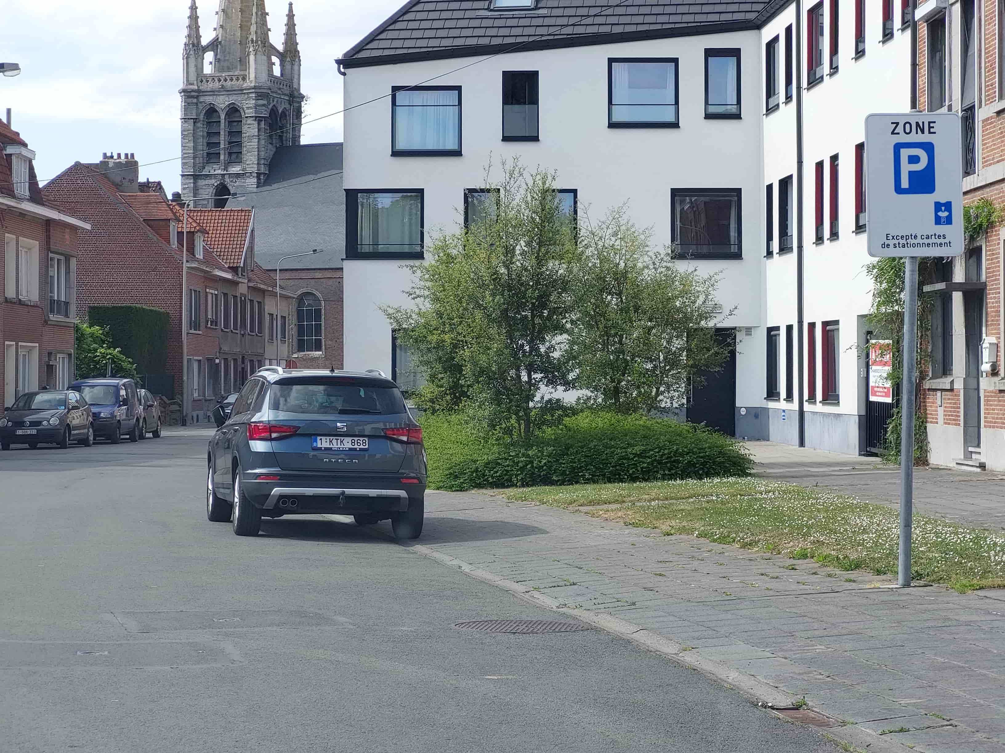 Absolument ! En tant qu'expert du code de la route belge, voici une description détaillée de la situation routière présentée dans l'image.
Description Détaillée de la Situation Routière pour l'Examen de Conduite
La scène se déroule sur une rue urbaine à sens unique ou à double sens non matérialisé, bordée de bâtiments résidentiels. L'environnement suggère un quartier de ville ou de petite ville en Belgique. Le temps est clair, avec un ciel partiellement nuageux, et il fait jour.
Type de situation routière :
Il s'agit d'une section de route droite dans un environnement urbain, sans intersection immédiate visible devant le véhicule du point de vue de l'observateur.
Sur la droite de la chaussée se trouve un trottoir pavé, bordé d'un caniveau et d'une bande de verdure avec de l'herbe et des petits arbres/arbustes, avant d'atteindre les façades des bâtiments.
Sur la gauche, des bâtiments résidentiels en briques bordent directement la chaussée, avec des véhicules stationnés en parallèle.
Éléments visibles :
Véhicules :
Au premier plan, sur la droite, une SUV de couleur gris foncé (Seat Ateca), immatriculée en Belgique (plaque 1-KTK-868), est stationnée ou à l'arrêt, le derrière orienté vers l'observateur. Ses roues droites sont très proches du bord de la chaussée (le caniveau), et ses roues gauches sont sur l'asphalte, laissant une partie de la voie libre.
Sur le côté gauche de la rue, plusieurs véhicules sont stationnés en parallèle le long du trottoir. Un véhicule noir (qui semble être une VW Polo, immatriculée 1-GQX-173) est clairement visible, suivi d'une camionnette bleue et d'une autre voiture grise.
Panneaux de signalisation :
Le panneau le plus proéminent est situé sur la droite, juste après le véhicule SUV. C'est un panneau de zone de stationnement réglementé (signal F1a "ZONE P") de forme rectangulaire, blanc avec une bordure bleue.
En haut, il porte l'inscription "ZONE" en majuscules.
En dessous, un grand "P" bleu sur fond blanc, symbolisant le stationnement (signal E9a).
Plus bas, en lettres noires, la mention "Excepté cartes de stationnement" est visible, accompagnée d'un petit pictogramme montrant une carte insérée dans un horodateur ou un parcmètre.
Feux : Aucun feu de signalisation (rouge, orange, vert) n'est visible. Des lampadaires sont présents mais non pertinents pour la question.
Marquages au sol : La chaussée est asphaltée. Aucun marquage au sol spécifique lié au stationnement (lignes jaunes, bleues, délimitation de places) n'est clairement visible sur l'asphalte devant le panneau ou autour du SUV. Cela indique que la réglementation de stationnement est principalement dictée par la signalisation verticale (le panneau).
Bâtiments et environnement :
À droite, juste derrière le panneau, se trouve un bâtiment moderne à la façade blanche avec des encadrements de fenêtres noirs. Plus loin sur la droite, on aperçoit des bâtiments en briques plus anciens.
À gauche, une série de maisons en briques traditionnelles belges, avec un clocher d'église en pierre grise (type gothique ou néo-gothique) visible au loin, au-dessus des toits.
De la végétation (arbres et arbustes) est présente sur la bande de verdure à droite, ainsi que de l'herbe avec de petites fleurs blanches.
Position et direction des véhicules :
Le SUV gris foncé est positionné sur la droite de la chaussée, orienté dans le même sens que le panneau (éloignement de l'observateur). Il est garé longitudinalement.
Les voitures sur la gauche sont également garées longitudinalement, en parallèle, le long du trottoir de gauche.
Panneaux de signalisation présents et leur signification :
Panneau F1a "ZONE P" avec mention "Excepté cartes de stationnement" et pictogramme :
Ce panneau indique l'entrée dans une zone de stationnement réglementé. Dans cette zone, des règles spécifiques de stationnement s'appliquent à tous les usagers de la route.
La mention "Excepté cartes de stationnement" signifie que le stationnement dans cette zone est généralement payant ou limité dans le temps (nécessitant un disque bleu ou un ticket d'horodateur), SAUF pour les véhicules munis d'une carte de stationnement spécifique (par exemple, carte de riverain, carte professionnelle, ou la carte de stationnement pour personnes handicapées si elle est reconnue pour ces exemptions spécifiques). Pour les autres conducteurs, un horodateur ou un disque est requis, selon la réglementation locale précise de cette zone P.
Autre élément pertinent pour comprendre la situation :
Le contexte temporel fourni par la question : "Samedi 7 juillet à 13h." Ce détail est crucial car les règles de stationnement (notamment les périodes payantes ou de zone bleue) varient souvent en fonction du jour de la semaine et de l'heure. Il est fréquent que le stationnement soit gratuit le samedi après-midi dans certaines zones, mais ce n'est pas systématique et dépend de l'arrêté communal spécifique à la zone P.
En résumé, le conducteur est sur le point d'entrer dans une zone où le stationnement est réglementé et potentiellement payant ou limité dans le temps, sauf pour les détenteurs de cartes spécifiques. La question posée concerne la possibilité de se stationner sur la droite au-delà* de ce signal, en tenant compte du jour et de l'heure.