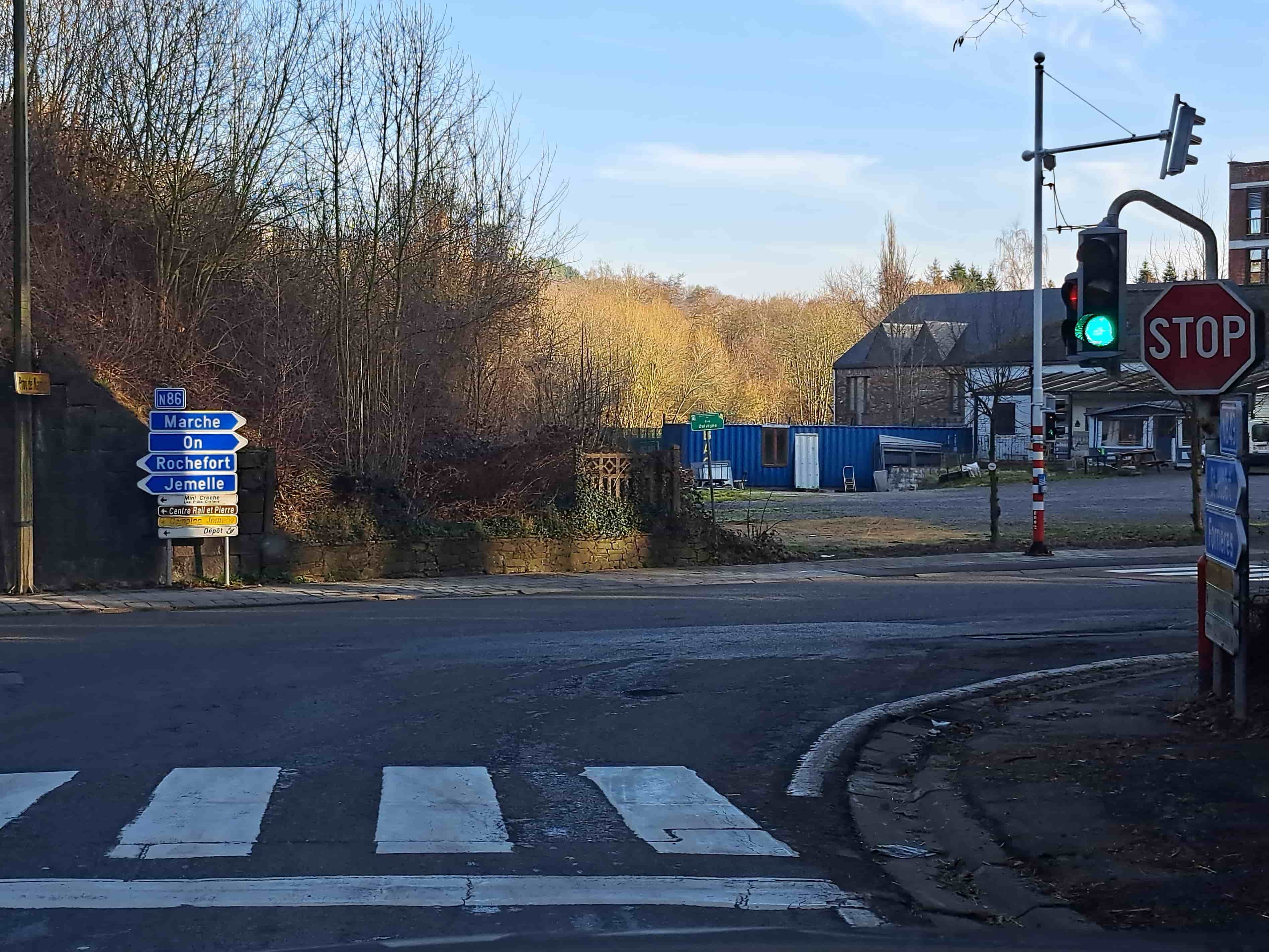 Excellent! En tant qu'expert en code de la route belge, je vais analyser cette image point par point pour fournir une description détaillée.
Description Détaillée de la Situation Routière (Belgique)
L'image présente une situation routière complexe et courante dans le cadre d'un examen de conduite en Belgique, focalisant sur l'interaction entre les feux de circulation et les panneaux de signalisation.
Type de situation routière:
Il s'agit d'une intersection complexe (carrefour) située probablement en zone périurbaine ou à l'entrée d'une localité. Le point de vue est celui d'un conducteur s'approchant de cette intersection. La présence de multiples panneaux de direction indique un carrefour permettant d'accéder à plusieurs localités et points d'intérêt. La route principale semble être la N86.
Éléments visibles:
Marquages au sol:
Au premier plan, un passage pour piétons (zébra) est clairement visible, marquant l'endroit où les piétons peuvent traverser la chaussée.
Immédiatement après le passage pour piétons et avant l'intersection principale, une ligne d'arrêt (ligne blanche continue) est tracée sur la chaussée.
Signalisation lumineuse (Feux de circulation):
Un feu de circulation pour véhicules est suspendu au-dessus de la voie, affichant une lumière verte. Ce feu est monté sur un poteau rayé rouge et blanc, souvent utilisé pour augmenter sa visibilité.
Un autre feu de circulation est visible en hauteur sur le mât principal, mais son état n'est pas clairement discernable.
Panneaux de signalisation verticaux:
Panneau STOP (B5): Un grand panneau octogonal rouge avec le mot "STOP" en blanc est situé juste à côté et légèrement en dessous du feu de circulation affichant le vert. Sa présence simultanée avec un feu vert est l'élément central de la question.
Panneaux de direction (F1a): Un ensemble de panneaux bleus avec des flèches blanches indique les directions pour "Marche", "On", "Rochefort", et "Jemelle", avec l'indication de la route N86. En dessous, des panneaux plus petits à fond jaune et flèches noires indiquent des points d'intérêt locaux tels que "Mini Crèche", "Centre Rail et Pierre", "Camping Jemelle", et "Dépôt".
Un petit panneau vert, visible sur la droite avant les conteneurs, indique "Rue Delvigne".
Un panneau partiellement masqué à gauche du mur pourrait être un panneau de propriété privée.
Environnement et éléments du paysage:
À gauche de la route, un talus boisé et une végétation dense (arbres sans feuilles, probablement fin d'hiver/début printemps) limitent la visibilité.
À droite, la visibilité est également entravée par des bâtiments, des conteneurs bleus et d'autres structures qui bordent la route ou la propriété adjacente.
Un bâtiment en pierre de couleur foncée se distingue en arrière-plan à droite.
Le ciel est dégagé, bleu avec quelques nuages.
La chaussée est en asphalte. Un trottoir borde la route sur la droite.
Position et direction des véhicules s'il y en a:
Aucun véhicule n'est visible en mouvement immédiat dans l'intersection. La photo est prise du point de vue du conducteur qui s'apprête à entrer dans l'intersection. Un petit fourgon blanc est très partiellement visible plus loin sur la route à droite.
Panneaux de signalisation présents et leur signification:
Feu vert (signal lumineux): En Belgique, un feu vert signifie que le conducteur est autorisé à passer l'intersection, à condition de le faire en toute sécurité et de céder le passage aux piétons éventuellement engagés.
Panneau STOP (B5): Ce panneau ordonne au conducteur de s'arrêter obligatoirement avant la ligne d'arrêt et de céder le passage à tous les usagers venant des autres directions, avant de s'engager.
Panneaux de direction (F1a): Indiquent les directions vers différentes localités ou points d'intérêt.
Tout autre élément pertinent pour comprendre la situation:
Hiérarchie des signaux: C'est l'élément le plus crucial. En droit routier belge (et dans la plupart des pays européens), les signaux lumineux (feux de circulation) priment sur les signaux routiers permanents (panneaux de signalisation). Si un feu de circulation fonctionne, c'est lui qui doit être obéi en priorité.
Visibilité réduite: La question mentionne "pas de visibilité à droite et à gauche". L'image confirme cette difficulté, avec un talus boisé à gauche et des bâtiments/conteneurs à droite qui masquent les vues latérales, rendant la situation potentiellement dangereuse si l'on ne respecte pas la primauté du feu.
• Le danger d'une signalisation contradictoire: La présence simultanée d'un feu vert et d'un panneau STOP est une source de confusion potentielle pour les conducteurs. Cependant, la règle de la hiérarchie est claire. Le panneau STOP est souvent laissé en place soit comme signalisation de secours en cas de panne des feux, soit pour réguler un mouvement spécifique (par exemple, un virage à droite en cas de feu rouge pour le trafic principal, mais ce n'est pas le cas ici).
Contexte de la question: "J'ai le feu vert mais je n'ai pas de visibilité à droite et à gauche, dois-je respecter le STOP ?"
La description détaillée ci-dessus met en évidence la contradiction entre le feu vert et le panneau STOP, ainsi que la visibilité réduite. La réponse à cette question réside dans la connaissance de la hiérarchie des signaux.