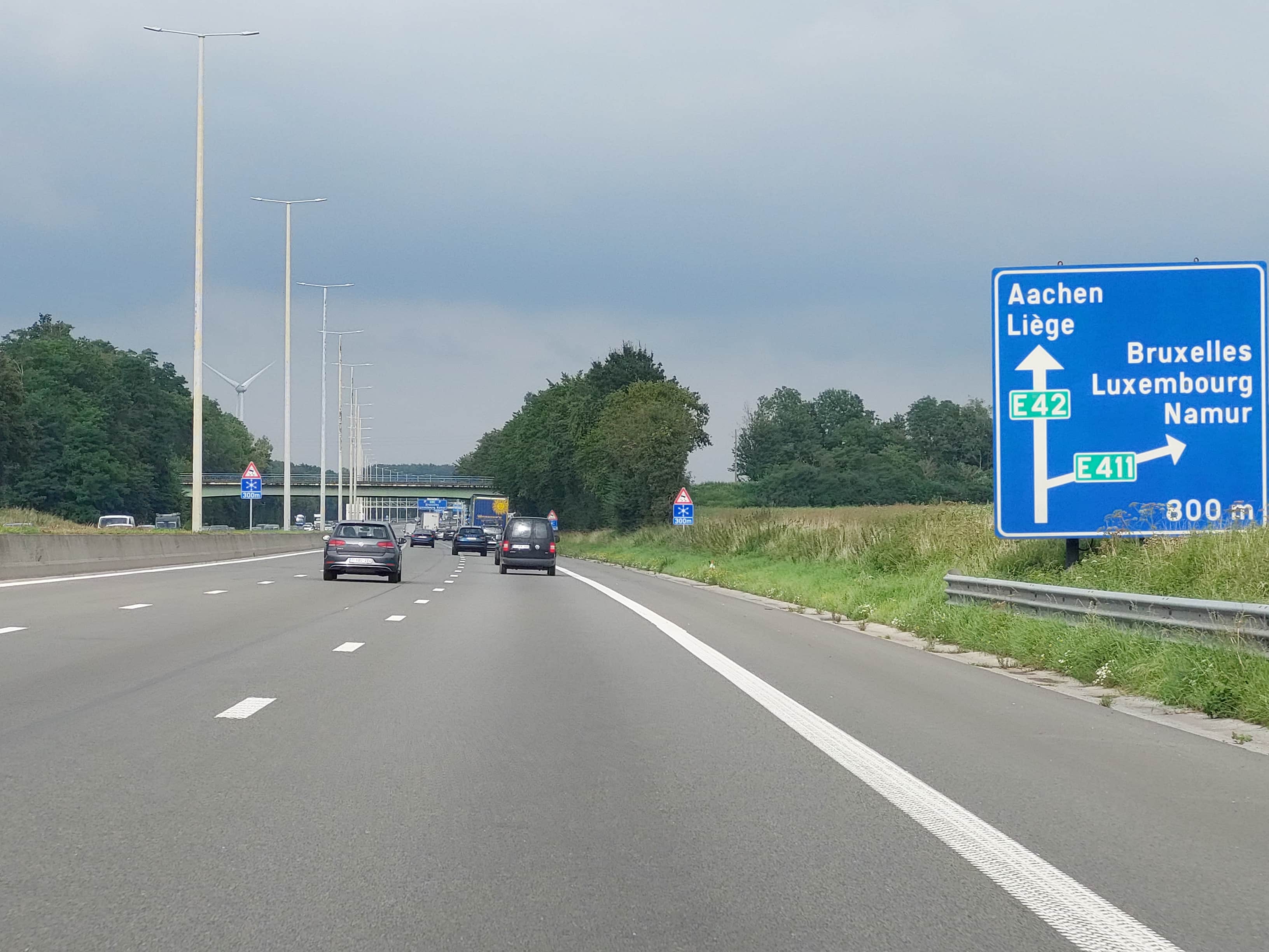En tant qu'expert en code de la route belge, voici une analyse détaillée de la situation routière présentée sur l'image :
Contexte de la question: "Actuellement, je circule sur :"
Le type de situation routière:
La scène se déroule sur une autoroute belge (ou voie assimilée) à chaussées séparées, avec plusieurs bandes de circulation dans le même sens. Le conducteur approche d'une bifurcation ou d'un échangeur autoroutier, comme l'indique la signalisation. Ce n'est pas une simple route ou intersection urbaine, mais une infrastructure majeure destinée au trafic rapide.
Les éléments visibles:
Chaussée: L'asphalte est en bon état. On distingue clairement au moins deux bandes de circulation dans le sens du conducteur, plus un accotement de droite (bande d'arrêt d'urgence) séparé par une ligne blanche continue à relief (bande rugueuse). Une glissière de sécurité en béton sépare la chaussée du terre-plein central à gauche, indiquant des voies de circulation opposées.
Marquages au sol: Des lignes discontinues blanches séparent les bandes de circulation. Une ligne continue blanche avec un relief "bande rugueuse" délimite l'accotement de droite.
Véhicules: Plusieurs véhicules circulent devant le conducteur, tous dans le même sens. On aperçoit notamment une voiture grise (type VW Golf) sur la bande de gauche, une voiture foncée sur la bande du milieu et une camionnette noire sur la bande de droite, ainsi qu'un poids lourd plus loin. Le trafic semble modéré.
Infrastructure environnante:
De nombreux lampadaires sont alignés le long de l'autoroute, suggérant un éclairage de nuit.
Un pont ou viaduc enjambe l'autoroute plus loin dans la perspective, indiquant potentiellement un autre axe routier.
Une végétation dense (arbres, buissons) borde l'autoroute, séparant la route du paysage de champs et de prairies.
Une éolienne est visible en arrière-plan, à travers les arbres, sur la gauche.
La position et direction des véhicules s'il y en a:
Tous les véhicules visibles circulent dans la même direction que le véhicule du conducteur (qui prend la photo). Ils sont positionnés sur les différentes bandes de circulation, respectant a priori les règles de circulation (ex: le véhicule le plus à droite est une camionnette, potentiellement plus lente). Aucun véhicule ne semble être en phase de manœuvre (changement de bande, signal d'une sortie) à ce stade.
Les panneaux de signalisation présents et leur signification:
Grand panneau bleu rectangulaire (à droite de la chaussée) :
Il s'agit d'un panneau de pré-signalisation d'un échangeur autoroutier (Panneau de type E9a en Belgique).
Il indique une bifurcation à 800 mètres.
La flèche vers le haut (tout droit) indique la direction vers Aachen (Allemagne) et Liège, sur l'autoroute E42.
La flèche oblique vers la droite indique une sortie vers Bruxelles, Luxembourg et Namur, via l'autoroute E411.
Ce panneau informe le conducteur des destinations et des numéros d'autoroutes, lui permettant de choisir sa bande de circulation à l'avance.
Petit panneau de danger triangulaire (sur la gauche, avant le pont) :
C'est un panneau de danger A23 : "Chaussée glissante" (pictogramme de flocon de neige sur fond blanc bordé de rouge).
Il est complété par un panneau additionnel rectangulaire bleu indiquant "300 m".
Signification : Il avertit le conducteur qu'à 300 mètres, il y aura une section de chaussée potentiellement glissante, souvent due au verglas ou à la neige, notamment sur les ponts ou viaducs comme celui visible au loin. Le conducteur doit donc s'attendre à une adhérence réduite et adapter sa conduite en conséquence.
Tout autre élément pertinent pour comprendre la situation:
La présence des numéros d'autoroutes européens (E42, E411) confirme qu'il s'agit bien d'un réseau autoroutier.
Le panneau de pré-signalisation à 800m est crucial : il signifie que la décision de prendre la sortie E411 ou de continuer sur l'E42 doit être anticipée, mais le point de divergence physique n'est pas encore atteint. Le conducteur circule actuellement sur le tronc commun de l'autoroute avant la séparation.
• Le panneau de danger A23 concernant la chaussée glissante, même s'il ne semble pas y avoir de conditions hivernales au moment de la photo (ciel nuageux mais pas de neige/gel), est un rappel de vigilance typique sur ces infrastructures, particulièrement sur les ponts.
En résumé, le conducteur circule actuellement sur une autoroute belge, en approche d'un échangeur important qui propose deux directions principales (Aachen/Liège via E42 ou Bruxelles/Luxembourg/Namur via E411 à 800m), et est averti d'une section potentiellement glissante à 300m.