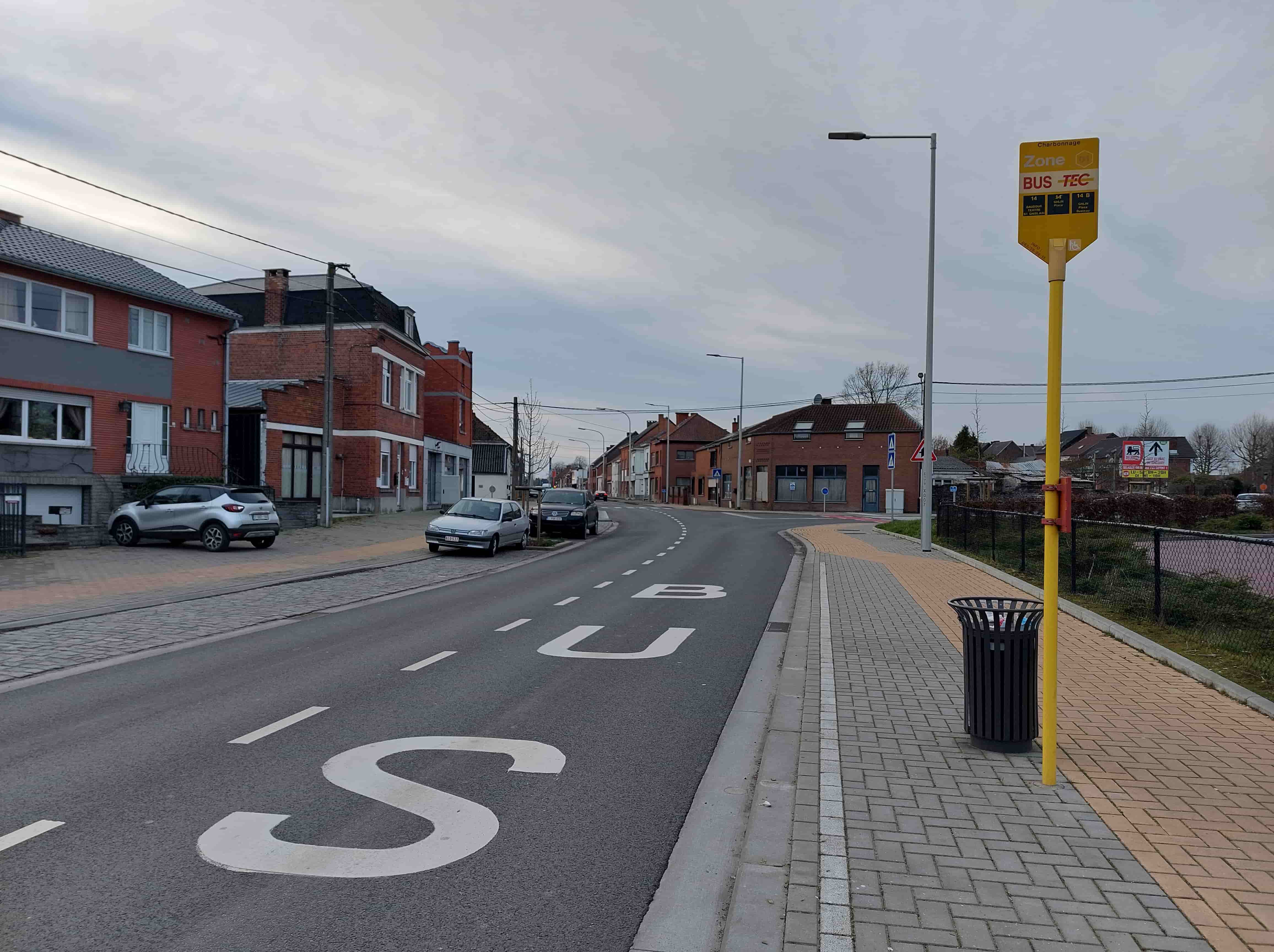 Absolument ! Voici une description détaillée de la situation routière présente sur l'image, en tant qu'expert du code de la route belge, pour comprendre le contexte de la question posée.
Description détaillée de la situation routière
L'image présente une vue de face d'une section de route bidirectionnelle dans ce qui semble être une agglomération ou une zone résidentielle en Belgique. Le temps est couvert.
Type de situation routière:
Il s'agit d'une section droite d'une chaussée publique, bordée de chaque côté par des habitations. Sur la droite, le trottoir est large et présente une zone spécifiquement aménagée pour un arrêt de bus. Plus loin sur la droite, la route semble s'incurver légèrement vers la droite et il y a une intersection ou un carrefour plus en avant, signalé par d'autres panneaux.
Éléments visibles:
Chaussée: Revêtue d'asphalte gris foncé.
Marquages au sol: Des lettres blanches, épaisses et bien visibles, épellent le mot "BUS" sur la chaussée, juste avant l'emplacement du poteau d'arrêt de bus. Plus à gauche, des lignes pointillées blanches délimitent probablement la zone de stationnement ou le bord de la voie de circulation.
Trottoir: Sur la droite, un trottoir large et bien entretenu, pavé de dalles de béton. La zone directement sous le panneau d'arrêt de bus et légèrement avant est pavée de dalles de couleur beige-jaune, contrastant avec les dalles grises adjacentes, ce qui est une pratique cournante pour signaler visuellement l'emplacement de l'arrêt.
Panneau d'arrêt de bus (L40): Un poteau jaune vif supporte un panneau rectangulaire également jaune portant les inscriptions "Charbonnage", "Zone 01", et en grand "BUS - TEC". Il indique les lignes de bus (14, 14A, 14B) et leurs destinations, ainsi qu'un pictogramme d'accès pour personne à mobilité réduite. Ce panneau est une signalisation d'arrêt d'autobus, un panneau L40.
Corbeille publique: Une poubelle métallique de couleur anthracite est fixée au poteau de l'arrêt de bus.
Éclairage public: Un lampadaire de rue est visible juste derrière le panneau de l'arrêt de bus. D'autres lampadaires sont également visibles le long de la rue.
Bâtiments: Des maisons d'habitation en briques rouges et grises bordent la route des deux côtés, certaines avec des entrées de garage ou des allées privées.
Véhicules:
Sur la gauche, garée dans une entrée de propriété (partiellement sur un trottoir pavé), se trouve une voiture de type SUV compacte gris clair (Renault Captur).
Plus loin sur la gauche, stationnées le long de la chaussée, se trouvent une voiture citadine gris clair (Peugeot 306) et, derrière elle, un SUV foncé (possiblement Volkswagen).
Une autre voiture est visible très loin sur la chaussée, au fond de l'image.
Clôture et végétation: Sur la droite, après le trottoir, une clôture grillagée délimite un terrain qui semble être en friche ou en cours d'aménagement, avec quelques arbustes.
Autres panneaux (plus loin sur la droite): On aperçoit, près de l'intersection lointaine, un panneau de danger (triangle rouge avec point d'exclamation, A1) et un panneau carré bleu avec deux flèches verticales blanches (F19), qui indique une traversée de piétons ou une traversée pour cyclistes à proximité. Il y a aussi des panneaux commerciaux (Delhaize).
Position et direction des véhicules s'il y en a:
L'image est prise depuis le côté droit de la chaussée, juste avant les marquages "BUS". Le conducteur qui poserait la question serait donc situé à cet endroit, se dirigeant vers le fond de l'image. Les voitures garées sur la gauche sont stationnées dans le même sens de circulation que le conducteur imaginaire.
Panneaux de signalisation présents et leur signification:
Panneau L40 ("BUS - TEC"): C'est le panneau principal ici. Il indique un arrêt d'autobus. En Belgique, le Code de la route stipule qu'il est interdit de stationner (immobilisation prolongée) à moins de 15 mètres de part et d'autre de ce panneau. Il est également interdit de s'arrêter (immobilisation le temps nécessaire pour embarquer ou débarquer des personnes ou des choses) à l'emplacement où les autobus prennent ou déposent des voyageurs.
Marquage au sol "BUS": Ce marquage renforce la signalisation de l'arrêt de bus et délimite visuellement la zone où l'arrêt des bus est prévu.
Panneau A1 (triangle rouge danger) et F19 (traversée piétons/cyclistes): Ces panneaux, situés plus loin sur la droite, signalent un danger général et une traversée de piétons/cyclistes à venir, mais ne sont pas directement pertinents pour la zone immédiate de l'arrêt de bus.
Tout autre élément pertinent pour comprendre la situation:
La question fait la distinction cruciale entre "s'arrêter quelques secondes" (arrêt) et "stationner" (parking). Le fait de vouloir "décharger un ami" est un motif d'arrêt, non de stationnement. L'absence de lignes jaunes (continues ou discontinues) le long du trottoir à cet endroit est également à noter, bien que la signalisation de l'arrêt de bus ait sa propre réglementation spécifique.