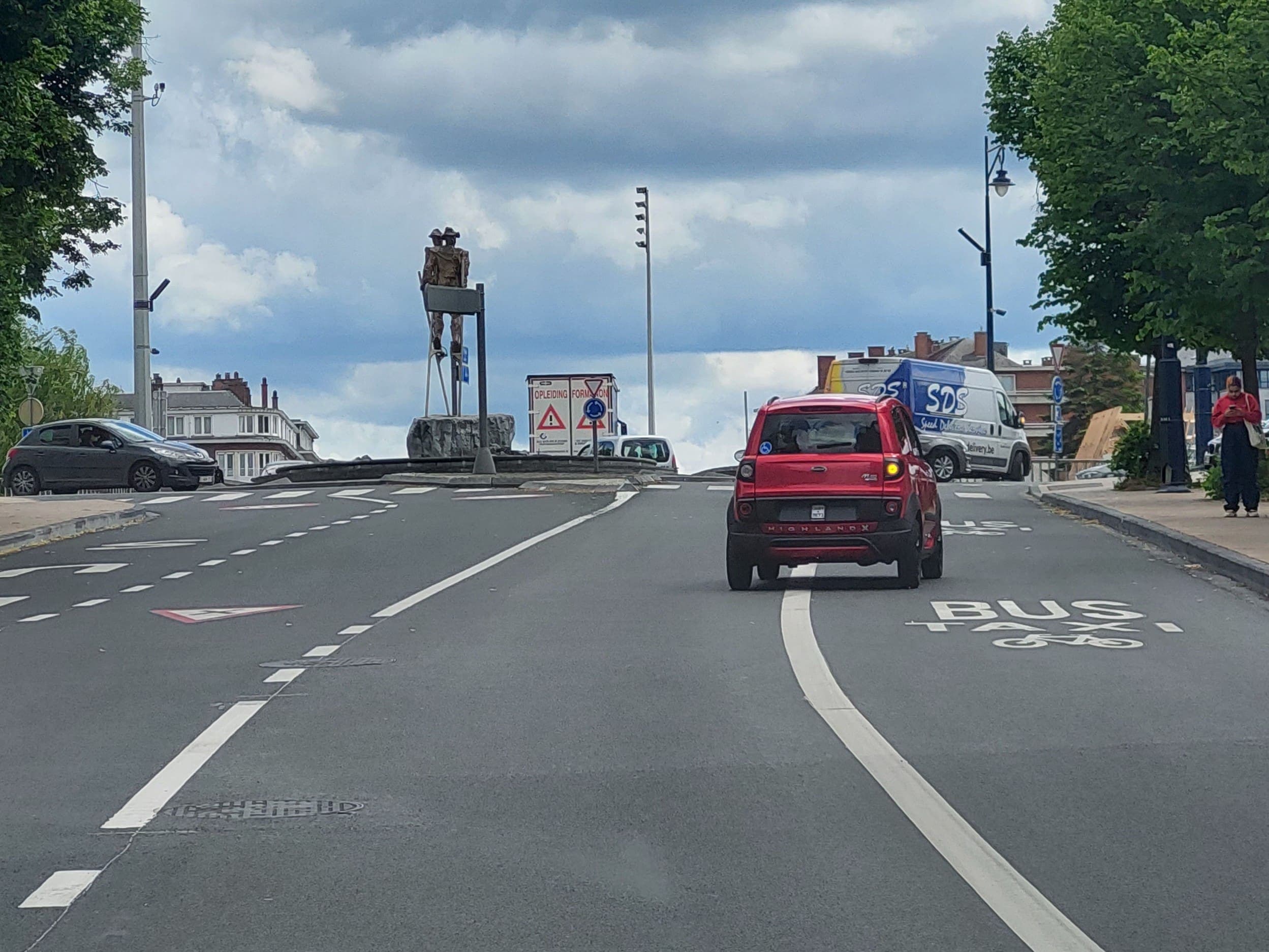 Absolument ! En tant qu'expert en code de la route belge, je vais analyser cette image pour vous fournir une description détaillée.
Description détaillée de la situation routière :
Type de situation routière :
Nous nous trouvons à l'approche d'un carrefour à sens giratoire (communément appelé rond-point en Belgique). La chaussée est une route à plusieurs voies menant à ce rond-point. Il s'agit d'un environnement urbain ou périurbain, avec des bâtiments en arrière-plan et de la végétation sur les côtés.
Éléments visibles :
Le véhicule du conducteur (implicite) : L'image est prise depuis l'intérieur d'un véhicule, qui se trouve dans la voie de droite, derrière le véhicule rouge.
Le véhicule rouge : Il s'agit d'une petite voiture rouge (type voiturette ou micro-car, potentiellement une Aixam Highland X, comme indiqué à l'arrière) positionnée directement devant le véhicule du conducteur. Son clignotant droit est activé, indiquant son intention de tourner à droite. Un autocollant bleu représentant une personne en fauteuil roulant est visible à l'arrière, indiquant qu'il s'agit d'un véhicule utilisé par ou transportant une personne à mobilité réduite. Le véhicule rouge se trouve dans la voie la plus à droite.
Autres véhicules :
Une voiture foncée (gris anthracite, type berline compacte comme une Peugeot 207) est visible sur la gauche de la photo, arrêtée ou stationnée, semblant venir d'une voie adjacente ou être sur l'îlot central.
Une camionnette utilitaire blanche et bleue (marquée "SDS Speed Delivery.be") est visible sur la droite, plus loin dans le carrefour ou sur une voie de sortie.
Piéton : Une personne est visible sur le trottoir à l'extrême droite, marchant ou s'arrêtant, le regard fixé sur un téléphone portable.
Position et direction des véhicules :
Le véhicule du conducteur est en approche du rond-point, dans la voie de droite.
Le véhicule rouge est juste devant, dans la même voie de droite, et s'apprête à entrer dans le rond-point pour tourner à droite.
Panneaux de signalisation présents et leur signification (Code de la route belge) :
Panneaux verticaux (sur l'îlot central) :
Panneau C3 (rond bleu avec trois flèches blanches circulaires) : Indique un carrefour à sens giratoire. Il impose l'obligation de contourner l'îlot central par la droite.
Panneau B1 (triangle rouge inversé sur fond blanc) : Un panneau "Cédez le passage". Il indique que les conducteurs venant de cette direction doivent céder le passage à tous les véhicules circulant sur le rond-point.
Panneau A13 (triangle rouge avec point d'exclamation sur fond blanc) : Un panneau de danger général. Un panneau additionnel est visible en néerlandais "OPLEIDING FORM...", suggérant "Opleiding Formation" (formation), indiquant potentiellement une zone où se déroulent des activités de formation (par exemple, pour la conduite ou des travaux). La pertinence directe de ce panneau pour la manœuvre de virage est faible, mais il fait partie du décor.
Panneaux verticaux (sur la droite, partiellement cachés) :
Un panneau circulaire bleu avec une flèche blanche pointant vers le haut ou la droite est visible en arrière-plan à droite, mais trop obstrué pour être identifié avec certitude (pourrait être un sens unique D1 ou une direction obligatoire).
Marquages au sol :
"BUS TAXI" et symbole vélo : Le marquage au sol dans la voie de droite indique que cette voie est réservée aux bus, aux taxis et aux cyclistes.
Flèche de direction droite : Une grande flèche blanche peinte sur la chaussée, juste avant le marquage "BUS TAXI", indique que cette voie est destinée aux véhicules qui souhaitent tourner à droite.
Triangles "Cédez le passage" : Deux triangles blancs bordés de rouge sont peints sur la chaussée juste avant la ligne d'entrée du rond-point, renforçant l'obligation de céder le passage.
Ligne discontinue large : Sépare la voie "BUS TAXI" des autres voies.
Ligne discontinue (tirets) : Marque la ligne d'arrêt où le cédez le passage doit être respecté avant d'entrer dans le rond-point.
Tout autre élément pertinent pour comprendre la situation :
L'îlot central : Un îlot central imposant orné d'une sculpture (deux personnages sur des échasses) et de quelques arbustes structure le rond-point.
L'environnement : La scène se déroule sous un ciel nuageux mais la visibilité est bonne et la chaussée est sèche. Il s'agit d'une zone urbaine avec des habitations.
En résumé, vous êtes en approche d'un rond-point. Vous êtes, comme le véhicule rouge devant vous, dans une voie qui est explicitement signalée comme réservée aux bus, taxis et cyclistes, et une flèche au sol indique que cette voie mène à un virage à droite. L'entrée du rond-point est régie par un "Cédez le passage".
La question "Je souhaite suivre le véhicule rouge pour tourner à droite. Est-ce autorisé ?" porte principalement sur l'utilisation de cette voie réservée.