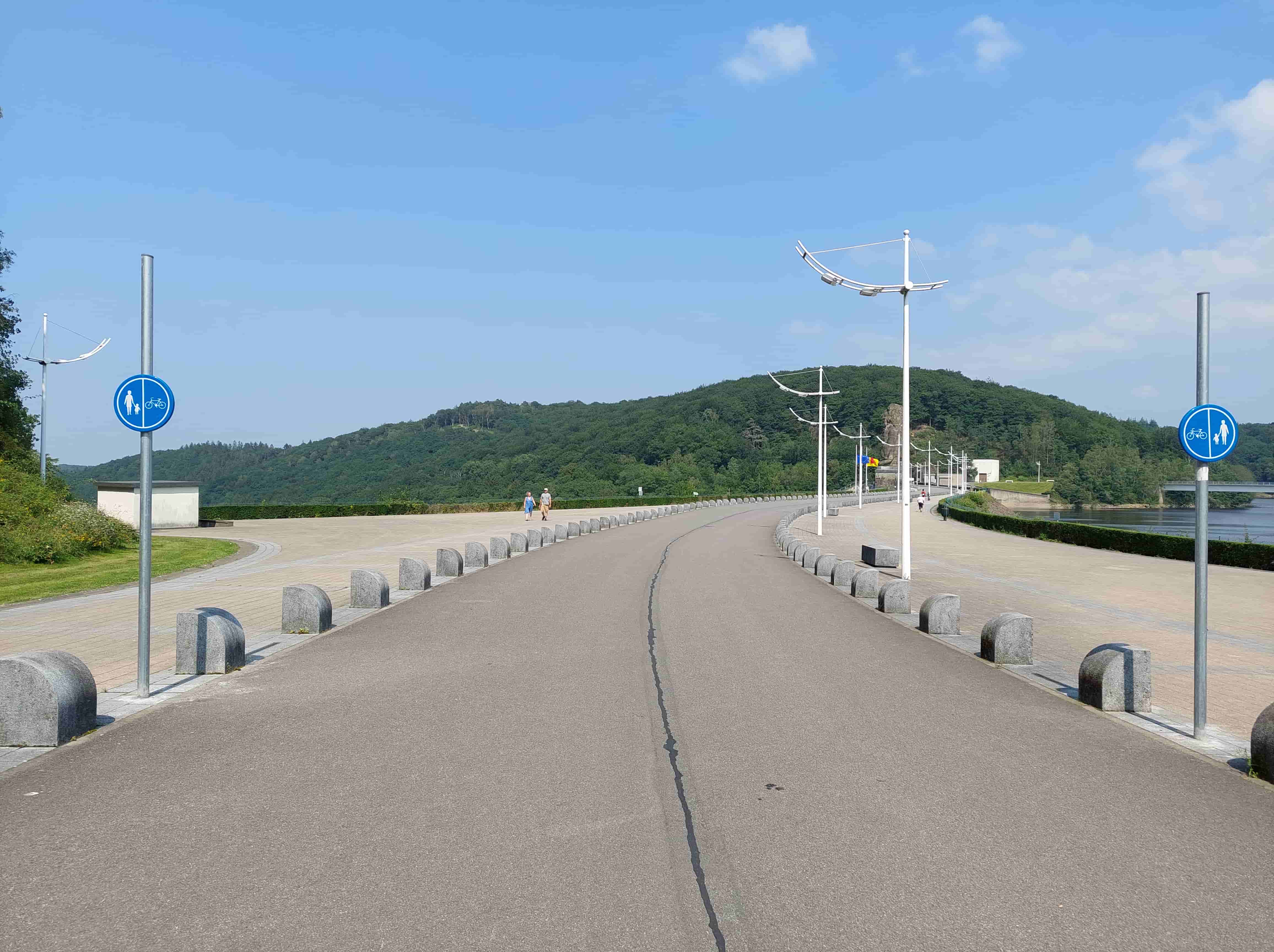 En tant qu'expert du code de la route belge, voici une description détaillée de l'image pour comprendre le contexte de la question :
Description Détaillée de la Situation Routière (Barrage de la Gileppe)
Type de situation routière:
L'image présente une portion du chemin qui traverse le couronnement (la crête) d'un barrage, identifiable comme le Barrage de la Gileppe par le contexte. Il s'agit d'une voie publique aménagée spécifiquement pour la promenade et la circulation douce, plutôt qu'une route destinée à la circulation générale des véhicules motorisés. Le chemin est légèrement courbé vers la droite dans la perspective de l'observateur.
Éléments visibles:
Chaussée/Chemin: Au premier plan et s'étendant vers l'horizon, on voit un chemin asphalté de couleur gris foncé, d'une largeur modeste. Une fine ligne plus foncée, qui semble être un joint de dilatation ou une démarcation, est visible au centre de cette bande d'asphalte.
Accotements/Zones latérales: De part et d'autre de cette bande asphaltée, il y a des zones plus larges et pavées (dalles de couleur claire), qui semblent être des trottoirs ou des zones de promenade.
Séparation: La bande asphaltée centrale est délimitée des zones pavées latérales par une série de bornes en béton ou en pierre de forme semi-cylindrique, espacées régulièrement. Ces bornes servent clairement à interdire le franchissement et à séparer les différents espaces de circulation.
Signalisation verticale: Deux panneaux de signalisation sont clairement visibles sur des poteaux métalliques.
Éclairage public: Plusieurs poteaux d'éclairage public au design moderne et épuré (avec des bras incurvés vers l'extérieur) sont alignés le long du chemin.
Végétation et paysages: En arrière-plan, on observe des collines boisées. Sur la gauche, un petit bâtiment blanc est partiellement visible. Sur la droite, on aperçoit une étendue d'eau (le lac du barrage) bordée d'une haie basse et de quelques arbres. Plus loin à droite, on distingue la silhouette du Lion de la Gileppe et des drapeaux.
Personnes: Deux personnes (un homme et une femme) marchent sur la zone pavée de gauche, s'éloignant de l'observateur. Une autre personne est visible plus loin sur la zone pavée de droite.
Absence de véhicules: Aucun véhicule motorisé (voiture, moto, camion) n'est visible sur la voie ou ses abords.
Position et direction des véhicules s'il y en a:
Il n'y a aucun véhicule motorisé visible sur l'image. Les personnes présentes sont des piétons, se déplaçant à pied le long des zones latérales pavées, dans le sens général du chemin.
Panneaux de signalisation présents et leur signification:
Deux panneaux identiques et de forme circulaire, à fond bleu avec des pictogrammes blancs, sont visibles sur des poteaux :
Un panneau est situé sur le côté gauche de la voie, au premier plan.
Le second panneau est visible sur le côté droit, plus loin sur le chemin.
Ces panneaux sont des signes D7 du Code de la Route belge : "Chemin obligatoire pour piétons et cyclistes".
Ils sont divisés verticalement : le côté gauche représente un pictogramme de piéton (un adulte tenant la main d'un enfant) et le côté droit représente un pictogramme de bicyclette.
Signification: Ce panneau indique une voie publique dont l'usage est réservé et obligatoire pour les piétons et les cyclistes. Dans ce cas précis, la division verticale du panneau D7 (a) ou D7 (b) signifie que les piétons doivent utiliser la partie du chemin signalée par leur symbole et les cyclistes celle signalée par le leur, mais l'absence de marquages au sol clairs pour séparer les deux parties de la chaussée pourrait prêter à confusion sur ce point. Cependant, la signification première et la plus importante pour la question est que cette voie est interdite à la circulation des autres véhicules (motorisés).
Tout autre élément pertinent pour comprendre la situation:
La présence des bornes en pierre le long du chemin renforce l'idée d'une restriction d'accès pour les véhicules de grande taille et d'une volonté de canaliser les usagers non motorisés.
Le cadre général (barrage, paysages, présence de piétons et de cyclistes implicite via les panneaux) suggère une zone récréative et touristique où la tranquillité et la sécurité des usagers faibles sont prioritaires.
• Le caractère asphalté du chemin central, sans marquages de voies automobiles, confirme qu'il n'est pas conçu pour la circulation automobile classique.
En résumé, l'image montre une voie de circulation sur le barrage de la Gileppe, explicitement signalée par des panneaux D7 comme étant un chemin réservé et obligatoire pour les piétons et les cyclistes, et de fait, interdit à la circulation des véhicules motorisés.
