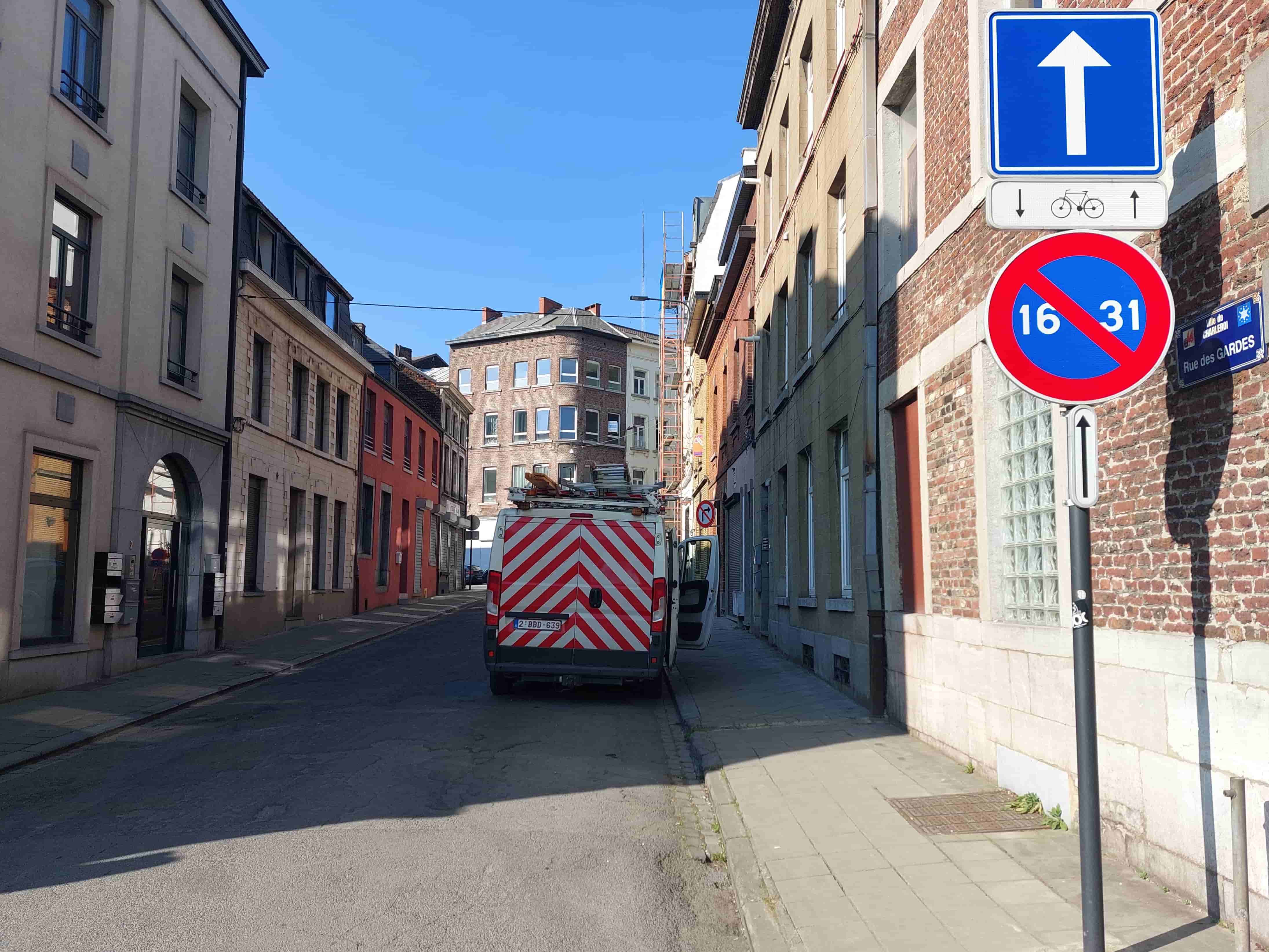Absolument ! En tant qu'expert du code de la route belge, je vais analyser cette image et la situation pour vous.
Description détaillée de la situation routière
Type de situation routière:
Nous sommes dans une rue urbaine relativement étroite, bordée de bâtiments résidentiels et commerciaux de plusieurs étages, typiques d'un centre-ville ou d'un quartier ancien. Il s'agit d'une rue à sens unique pour les véhicules motorisés, comme l'indique la signalisation. Des trottoirs sont présents des deux côtés de la chaussée.
Éléments visibles:
Chaussée: Le revêtement est goudronné et semble en bon état. Elle est suffisamment large pour permettre la circulation dans le sens unique.
Trottoirs: Des trottoirs pavés longent la rue des deux côtés. Celui de droite, où sont implantés les panneaux, est en bon état.
Bâtiments: Les bâtiments sont de styles variés, certains en briques rouges, d'autres en crépi. Ils sont rapprochés de la rue. Un échafaudage est visible sur un bâtiment plus loin sur la gauche.
Ciel: Le ciel est dégagé et d'un bleu clair intense, suggérant une journée ensoleillée. Des ombres portées distinctes sont visibles sur la droite (mur, poteau), indiquant une lumière du matin ou de fin d'après-midi.
Marquages au sol: Aucun marquage au sol particulier (lignes de stationnement, passages piétons, flèches directionnelles) n'est clairement visible sur la portion de route immédiate.
Panneau de nom de rue: Un petit panneau bleu sur le mur de droite indique "Ville de Charleroi - Rue des GARDES".
Position et direction des véhicules:
Véhicule au premier plan (ou juste après les panneaux): Un utilitaire (van) blanc avec des bandes obliques rouges et blanches à l'arrière (suggérant un véhicule d'intervention ou de chantier). Il porte une plaque d'immatriculation belge "2-BBD-639". Ce véhicule est stationné sur le côté gauche de la chaussée, dans le sens de la circulation indiqué par les panneaux (s'éloignant de l'observateur). Il est garé parallèlement au trottoir.
Véhicule au loin: Une voiture foncée est visible plus loin dans la rue, derrière le van, également dans le même sens de circulation.
Position de l'observateur: L'image est prise depuis le point de vue d'un conducteur arrivant dans la rue, juste avant les panneaux de signalisation qui lui font face.
Panneaux de signalisation présents et leur signification (Code de la route belge):
Les panneaux sont tous fixés sur un même poteau, côté droit de la chaussée :
Panneau supérieur (F19 - Rue à sens unique): Carré bleu avec une flèche blanche pointant vers le haut. Ce panneau indique le début d'une rue à sens unique dans la direction de la flèche. Tous les conducteurs de véhicules motorisés doivent suivre cette direction.
Panonceau sous le F19 (circulaire avec flèches et vélo): Panonceau blanc avec un pictogramme de vélo et deux flèches (une vers le bas, une vers le haut). Il s'agit du panneau additionnel M12 qui indique que la circulation à contresens est autorisée pour les cyclistes dans cette rue à sens unique.
Panneau inférieur (E1 + panonceau "16 31"): C'est un panneau d'interdiction de stationner (E1), rond avec un fond bleu barré de rouge. Sous ce panneau se trouve un panonceau rectangulaire blanc sur fond bleu avec les chiffres "16 31".
Le panneau E1 signifie "Stationnement interdit". Il interdit de laisser un véhicule à l'arrêt au-delà du temps nécessaire pour embarquer ou débarquer des personnes ou des choses, ou de laisser le véhicule sans conducteur.
Le panonceau "16 31" précise que cette interdiction de stationner est valable du 16 au 31 inclus de chaque mois. Cela implique que du 1er au 15 du mois, le stationnement serait autorisé sur ce côté de la chaussée (sauf autres interdictions).
Application des panneaux: Les panneaux F19 et M12 s'appliquent à l'ensemble de la chaussée à partir de leur implantation. Le panneau E1 avec le panonceau "16 31" s'applique au côté de la chaussée où il est implanté (le côté droit dans ce cas), et ce, à partir de ce point et jusqu'à la prochaine intersection, ou jusqu'à un panneau de fin d'interdiction.
Tout autre élément pertinent pour comprendre la situation:
La question contextuelle est : "Nous sommes le 23 juillet. Je veux arrêter mon véhicule au-delà de ces signaux routiers."
La date du 23 juillet est cruciale car elle tombe dans la période "16 31" mentionnée sur le panonceau de stationnement. Cela signifie que le stationnement est interdit sur le côté droit de la rue du 16 au 31 de chaque mois. Le 23 juillet, cette interdiction de stationnement est donc active sur le côté droit. L'arrêt, cependant, y reste autorisé s'il n'y a pas d'autres restrictions spécifiques. L'existence d'un van garé sur le côté gauche (où le panneau E1 ne s'applique pas directement) est également un élément visuel à noter.