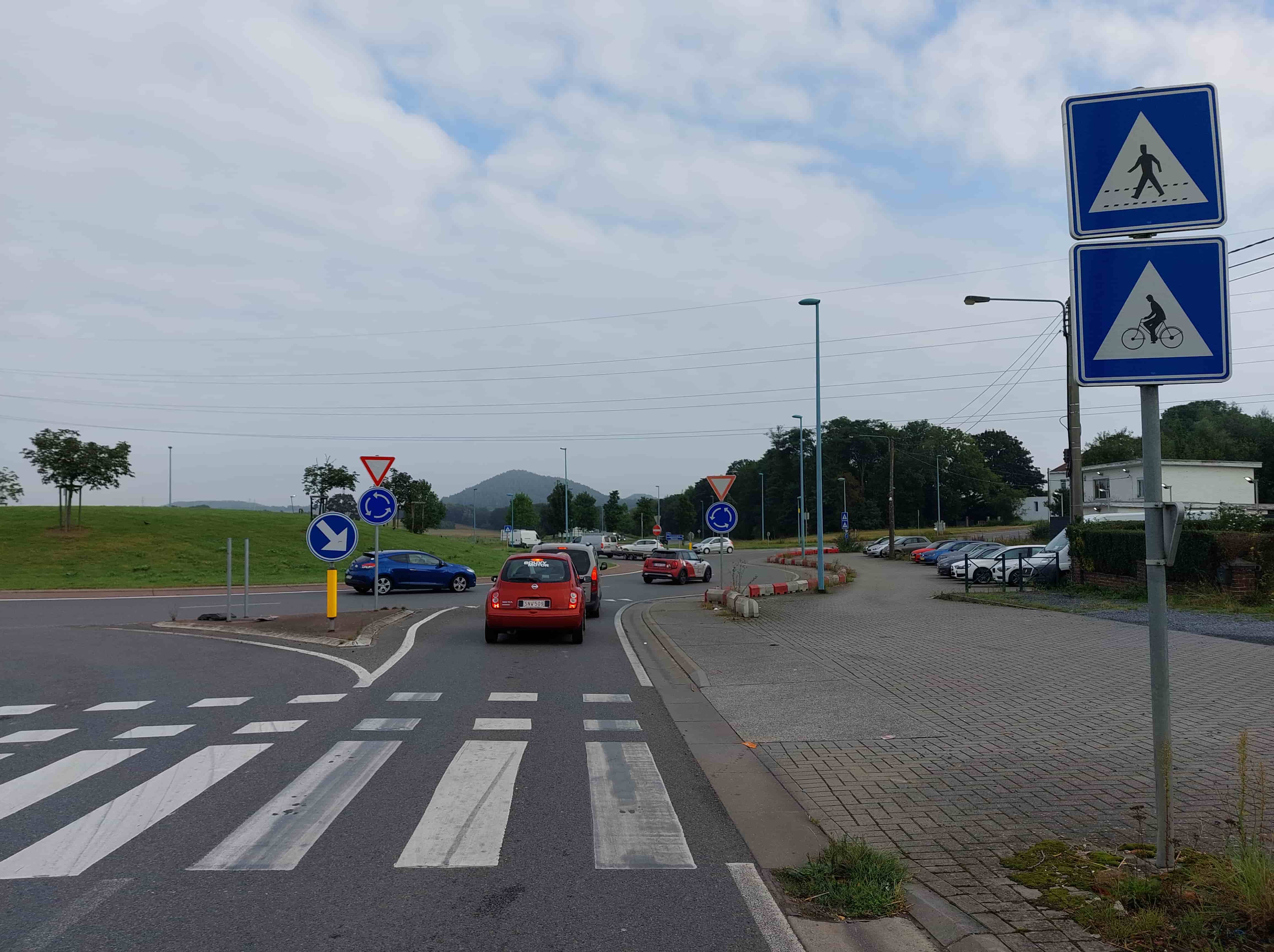 Voici une description détaillée de l'image, analysée par un expert en code de la route belge :
Description Détaillée de la Situation Routière
Type de situation routière :
La situation se présente comme l'approche d'un carrefour giratoire (rond-point). Le véhicule depuis lequel la photo est prise est sur une voie d'accès menant à ce rond-point. Il s'agit d'une zone où coexistent des flux de circulation automobile, cycliste et piétonne, typique des entrées d'agglomérations ou de zones commerciales/industrielles.
Éléments visibles :
La chaussée et les marquages au sol :
Devant le véhicule de la perspective (et la voiture rouge qui le précède), se trouvent clairement peints sur la chaussée :
Un passage pour piétons (marquage zebré avec de larges bandes blanches parallèles à l'axe de la route).
Immédiatement après le passage pour piétons, un passage pour cyclistes et conducteurs de cyclomoteurs à deux roues, reconnaissable aux carrés blancs peints sur la chaussée. C'est précisément à hauteur de ces carrés que la question est posée.
Plus loin, juste avant l'entrée du rond-point, une ligne discontinue de "cédez le passage" est visible.
Des lignes continues et discontinues délimitent les voies de circulation et l'îlot central du rond-point.
Véhicules :
Une voiture rouge (Nissan Micra) est arrêtée devant le véhicule de la perspective, positionnée au niveau des marquages au sol (passage piéton et cycliste).
Derrière la voiture rouge, un utilitaire gris clair est également visible.
À l'intérieur du rond-point, une voiture bleue (Renault Mégane coupé) est en circulation, se dirigeant de la gauche vers la droite de l'image.
Sur la droite de l'image, le long d'une voie d'accès ou d'un parking, plusieurs voitures sont garées (blanches, grises, bleues, rouges, etc.).
Panneaux de signalisation :
Sur l'îlot central de l'entrée du rond-point (à gauche de la voiture rouge) :
Un panneau B1 : "Cédez le passage" (triangle inversé blanc bordé de rouge).
En dessous, un panneau D5 : "Carrefour à sens giratoire" (rond bleu avec flèches blanches en sens anti-horaire).
En dessous encore, un panneau D1.a : "Direction obligatoire : passer à gauche de l'obstacle" (carré bleu avec une flèche blanche pointant vers le bas et la gauche).
Sur un poteau vertical à droite de la chaussée (au niveau des passages) :
Un panneau F.50 : "Passage pour piétons" (carré bleu avec pictogramme de piéton blanc sur un triangle blanc).
Juste en dessous, un panneau F.50bis : "Passage pour cyclistes et conducteurs de cyclomoteurs à deux roues" (carré bleu avec pictogramme de cycliste blanc sur un triangle blanc). Ce panneau signale la présence des carrés blancs au sol et est crucial pour la question.
Feux de signalisation :
Aucun feu de signalisation lumineux n'est visible.
Autres éléments :
Un îlot central gazonné pour le rond-point.
Des balises de délimitation rouges et blanches sur le bord droit de la chaussée, séparant la voie de circulation d'une aire de stationnement/accès.
Des lampadaires (poteaux bleus et gris foncé).
Des arbres, de la végétation, et un talus gazonné à gauche.
Des bâtiments en arrière-plan à droite.
Des lignes électriques aériennes.
Un ciel nuageux, suggérant une journée couverte mais une bonne visibilité générale.
Des collines ou montagnes en arrière-plan à l'horizon.
Position et direction des véhicules :
Le véhicule de la perspective est en approche de l'entrée du rond-point, sur la voie de droite, et est situé avant le passage piéton et le passage cycliste, probablement à l'arrêt ou ralentissant.
La voiture rouge devant est dans la même voie, à l'arrêt ou roulant très lentement au niveau des passages.
La voiture bleue circule dans le rond-point, venant de la gauche du point de vue de notre véhicule, et se dirigeant vers la droite.
Signification des panneaux de signalisation pertinents :
Panneau B1 "Cédez le passage" : Oblige le conducteur à céder le passage à tout véhicule venant de gauche ou de droite sur la chaussée abordée (dans le rond-point).
Panneau D5 "Carrefour à sens giratoire" : Indique la présence d'un rond-point où la circulation s'effectue dans le sens indiqué par les flèches.
Panneaux F.50 "Passage pour piétons" et F.50bis "Passage pour cyclistes et conducteurs de cyclomoteurs à deux roues" : Ces panneaux jumelés indiquent clairement la présence d'un passage où les piétons et les cyclistes/conducteurs de cyclomoteurs sont autorisés à traverser la chaussée. En Belgique, aux passages pour piétons, le piéton a priorité dès qu'il s'apprête à traverser ou est engagé. Pour les passages pour cyclistes et conducteurs de cyclomoteurs (marqués par les carrés blancs et le panneau F.50bis), ils ont priorité s'ils sont sur le point d'y traverser ou y sont engagés.
Autres éléments pertinents pour la question :
La question "A hauteur des carrés blancs peints sur la chaussée, dois-je céder le passage aux cyclistes et conducteurs de cyclomoteurs à 2 roues ?" fait directement référence au marquage au sol spécifique et au panneau F.50bis. Le conducteur est positionné de manière à aborder ces passages avant d'entrer dans le rond-point où un cédez le passage général s'applique aux véhicules motorisés. Le fait que la voiture rouge soit déjà arrêtée à ce niveau suggère une anticipation de cette priorité.