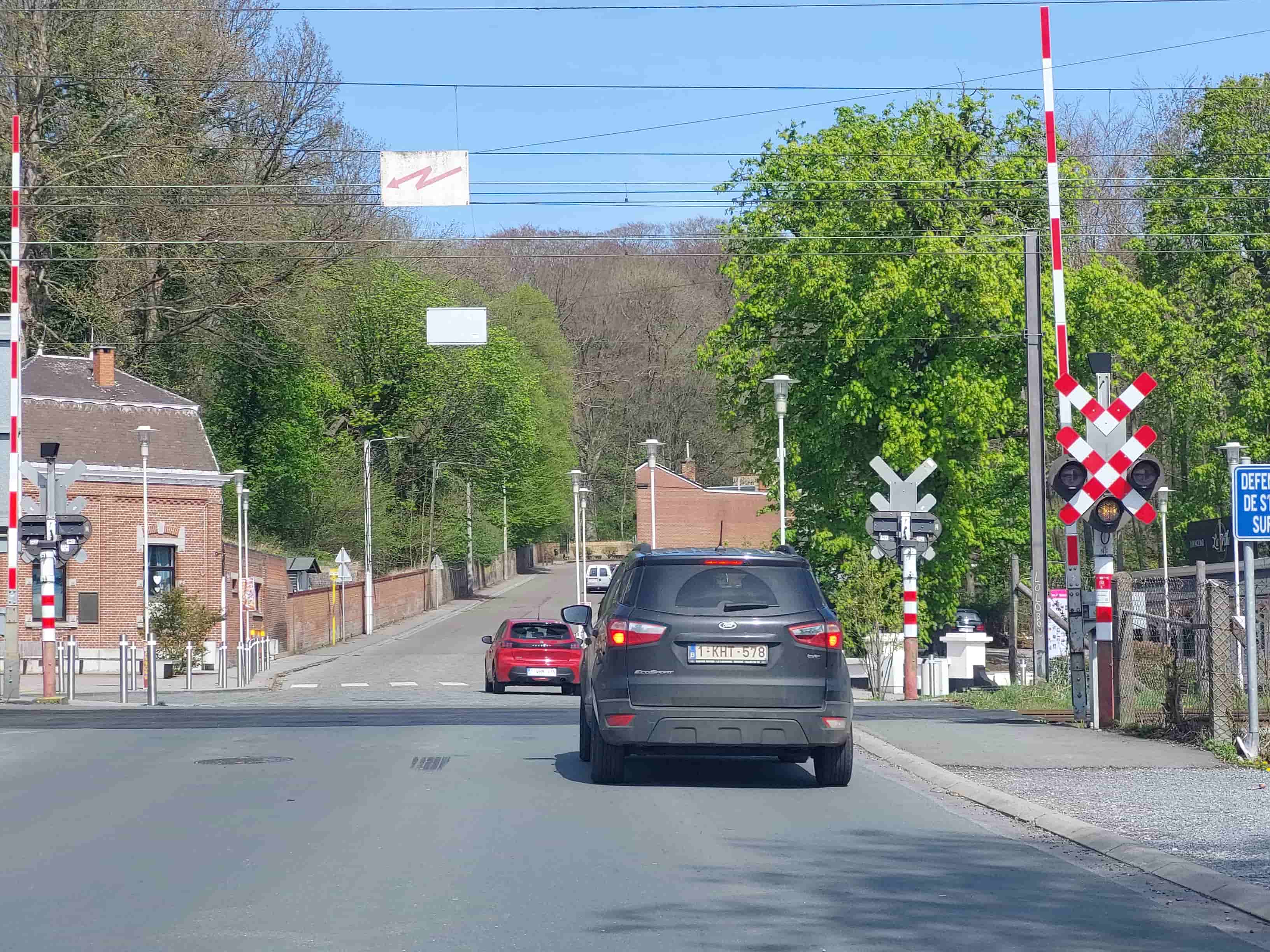 Absolument ! En tant qu'expert du code de la route belge, voici une analyse détaillée de l'image pour comprendre le contexte de la question :
Description détaillée de la situation routière
Type de situation routière:
La scène représente une section de route qui mène directement à un passage à niveau à barrières et feux lumineux. Il s'agit d'une route bidirectionnelle, avec une seule voie de circulation pour chaque sens (bien qu'il n'y ait pas de marquage central continu visible immédiatement après le véhicule devant, le passage à niveau en lui-même constitue une zone de danger). La route semble légèrement en pente montante après le passage à niveau, et est bordée d'arbres et de bâtiments, dont une ancienne gare ou maison de garde-barrière sur la gauche.
Éléments visibles:
Véhicules:
Le véhicule du conducteur est implicite, car la photo est prise de l'intérieur d'une voiture, regardant vers l'avant.
Une voiture de type SUV de couleur gris foncé (une Ford EcoSport, immatriculée 1-KHT-578) est juste devant le véhicule de l'observateur, avançant dans la même direction.
Plus loin, une voiture de couleur rouge (une Renault Clio, immatriculée 1-UHM-2xx) avance également dans la même direction, précédant le SUV gris.
Un véhicule utilitaire blanc (fourgonnette) est visible très loin sur la route, après le passage à niveau.
Passage à niveau:
Deux barrières de passage à niveau à rayures rouges et blanches sont clairement visibles : une partielle sur la gauche et une complète sur la droite. Elles sont actuellement en position ouverte (levée).
Un poteau de signalisation lumineuse est dressé à droite du passage à niveau.
Bâtiments et infrastructure:
Une bâtisse en briques de style ancien (suggérant une ancienne gare ou un bâtiment ferroviaire) est visible sur la gauche.
Des arbres et de la végétation dense bordent la route des deux côtés, avec des collines boisées en arrière-plan.
Plusieurs lampadaires sont présents le long de la route.
Des câbles électriques et/ou de caténaire traversent le ciel au-dessus de la route.
Position et direction des véhicules:
Les trois véhicules visibles (le SUV gris, la voiture rouge et le véhicule utilitaire lointain) circulent dans la même direction, s'approchant du passage à niveau. Le SUV gris est directement devant l'observateur. La voiture rouge est devant le SUV gris.
Panneaux de signalisation présents et leur signification (selon le Code de la route belge):
Signal lumineux de passage à niveau (avec Croix de Saint-André double et feux):
Une Croix de Saint-André double (panneau A 45) est fixée sur le poteau de signalisation lumineux à droite. Cette double croix indique qu'il s'agit d'un passage à niveau avec plusieurs voies ferrées.
Sous la Croix de Saint-André, on voit un ensemble de feux. Les deux feux rouges clignotants habituels sont présents mais ne semblent pas allumés.
TRÈS IMPORTANT : Un feu jaune intermittent est allumé sous les feux rouges. En Belgique, un feu jaune intermittent à un passage à niveau avec barrières signifie que le passage à niveau est en cours de fermeture. Il s'agit d'un signal d'avertissement immédiat indiquant qu'un train approche et que les barrières vont s'abaisser.
Barrières (ouvertes): Bien qu'actuellement levées, leur présence et le feu jaune intermittent annoncent leur fermeture imminente.
Panneau bleu rectangulaire (partiellement visible): Sur la droite, juste après le passage à niveau, un panneau bleu est visible et commence par "DÉFENSE DE S'ARRÊTER SUR...". Il s'agit probablement d'une interdiction de s'arrêter sur le passage à niveau, même en cas d'embouteillage, pour éviter d'être bloqué sur les voies.
Panneau blanc avec flèche en zig-zag rose: Un panneau blanc rectangulaire avec une flèche rose en forme de zig-zag pointant vers la gauche est suspendu au-dessus de la route. Ce n'est pas un panneau de signalisation routière standard, mais plutôt un indicateur lié aux installations ferroviaires, probablement en rapport avec les caténaires ou les lignes de contact.
Tout autre élément pertinent pour comprendre la situation:
Le détail le plus crucial pour répondre à la question de dépassement est l'état du feu jaune intermittent allumé au niveau du passage à niveau. Même si les barrières sont encore ouvertes et que les feux rouges ne clignotent pas encore, ce feu jaune signifie de manière formelle et sans équivoque que le passage à niveau est en cours de fermeture. Le contexte de la question mentionnant que la voiture devant avance à 5 km/h crée une tentation de dépasser, mais la signalisation actuelle est prioritaire.