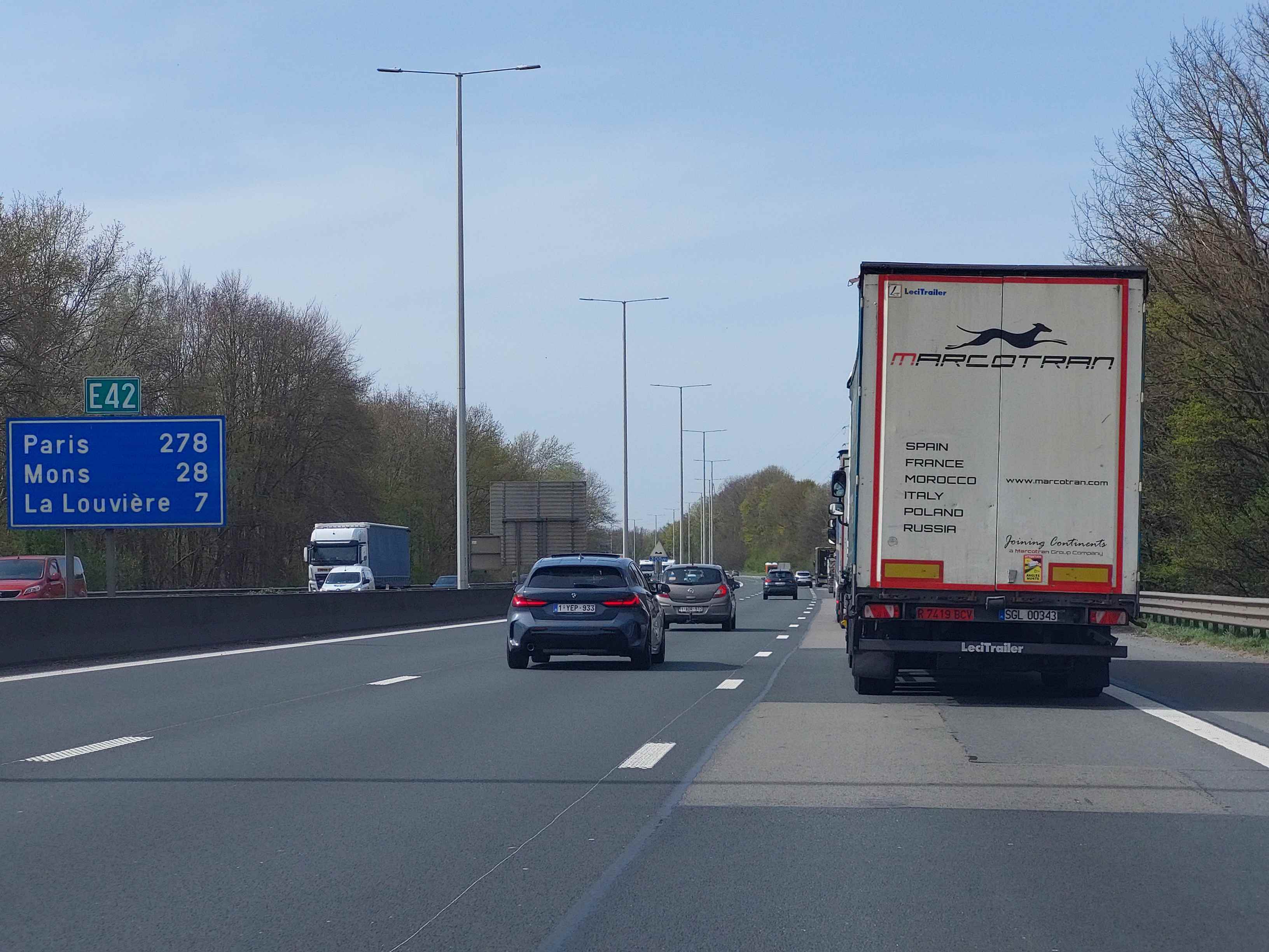 Absolument ! En tant qu'expert du code de la route belge, voici une description détaillée de la situation routière présentée dans l'image, en tenant compte du contexte de la question.
Description Détaillée de la Situation Routière (Examen de Conduite Belge)
L'image représente une scène sur une autoroute ou une voie rapide en Belgique, par une journée ensoleillée avec un ciel clair. La perspective est celle du conducteur d'un véhicule situé sur la voie de gauche (la voie de dépassement).
Type de situation routière:
Il s'agit d'une autoroute (ou voie rapide), caractérisée par une chaussée à plusieurs voies dans le même sens de circulation, séparée du sens inverse par un terre-plein central (bande médiane) et bordée par une bande d'arrêt d'urgence.
La route est rectiligne ou présente une très légère courbe vers la gauche au loin.
Aucune intersection ni feu de signalisation n'est visible.
L'environnement est rural ou périurbain, avec des massifs d'arbres et de la végétation dense le long des accotements de chaque côté de la chaussée.
Éléments visibles (véhicules, panneaux, feux, marquages au sol, etc.):
Chaussée:
Elle est composée d'au moins deux voies de circulation dans le sens de notre véhicule.
Une bande d'arrêt d'urgence est clairement visible sur la droite de la chaussée.
Les marquages au sol sont blancs et en bon état :
Une ligne discontinue large sépare la voie de gauche de la voie de droite.
Une ligne continue large sépare la voie de droite de la bande d'arrêt d'urgence, avec des bandes rugueuses (rumble strips) intégrées pour alerter les véhicules qui s'en approchent.
La voie de gauche est délimitée à son extrême gauche par une ligne continue qui jouxte la berme centrale (médiane).
Des catadioptres (yeux de chat) sont incrustés dans les marquages au sol.
Véhicules (dans notre sens de circulation):
Notre véhicule (point de vue): Se trouve sur la voie de gauche, prêt à dépasser.
Un semi-remorque: De marque "MARCOTRAN" (avec des mentions de pays européens comme Espagne, France, Italie, Maroc, Pologne, Russie) et "LeciTrailer". Il est situé juste à notre droite, sur la voie de droite, et semble avancer. Il porte un autocollant "ATTENTION ANGLES MORTS" à l'arrière.
Une voiture particulière (BMW Série 1, bleu foncé): Immatriculée "1-YEP-933", elle est devant notre véhicule sur la voie de gauche.
Autres véhicules: D'autres voitures sont visibles plus loin sur la voie de gauche derrière la BMW, et une autre (claire) est visible sur la voie de droite derrière le semi-remorque. L'ensemble du trafic semble dense mais fluide.
Véhicules (sens de circulation opposé):
Derrière la berme centrale, on aperçoit des véhicules circulant en sens inverse : un camion blanc, une camionnette blanche et une voiture rouge partiellement masquée par la glissière de sécurité.
Panneaux de signalisation:
Un grand panneau directionnel de couleur bleue est visible en hauteur sur le côté gauche de la chaussée. Il indique :
E42 (numéro d'autoroute européenne, encadré en vert).
Paris 278 (km)
Mons 28 (km)
La Louvière 7 (km)
Plus loin sur la droite, au niveau de l'accotement, un petit panneau d'indication ou de danger est visible, portant l'inscription "300m" sous un symbole triangulaire (trop lointain pour être identifié précisément, mais suggérant une zone de travaux ou un danger à venir).
Infrastructure et environnement:
Des lampadaires gris métalliques, dotés de doubles bras, sont régulièrement espacés le long de la berme centrale, indiquant que l'autoroute est éclairée la nuit.
Une berme centrale (médiane) en béton sépare les deux sens de circulation.
Des glissières de sécurité métalliques sont présentes le long des accotements.
La végétation est présente des deux côtés de l'autoroute, principalement des arbres sans feuilles (début de printemps ou fin d'automne).
Position et direction des véhicules:
Notre véhicule est dans la voie de gauche, le semi-remorque dans la voie de droite, la BMW devant nous dans la voie de gauche. Tous ces véhicules circulent dans le même sens, vers l'avant. Les autres véhicules visibles sont soit dans notre sens (devant nous), soit en sens inverse (au-delà de la berme centrale). La question indique notre intention de dépasser le semi-remorque, ce qui confirme notre position sur la voie de gauche.
Panneaux de signalisation présents et leur signification:
E42: Indique que nous nous trouvons sur l'autoroute européenne E42, qui traverse la Belgique (entre autres) et relie des villes importantes.
Panneau bleu directionnel (Paris, Mons, La Louvière): C'est un panneau de localisation et de distance (type F1a en Belgique). Il confirme que nous sommes bien en Belgique (villes de Mons et La Louvière) et nous donne la distance restante jusqu'à ces destinations.
Panneau "300m": Il s'agit probablement d'un panneau d'annonce de danger ou d'une zone particulière (ex: travaux, réduction de voie, bifurcation) à 300 mètres.
Tout autre élément pertinent pour comprendre la situation:
Le temps est clair et ensoleillé, offrant une excellente visibilité.
L'autocollant "ATTENTION ANGLES MORTS" sur le semi-remorque est un rappel important de la zone invisible pour le conducteur du camion, particulièrement pertinent lors d'une manœuvre de dépassement.
Le contexte de la question ("Je veux dépasser ce semi-remorque, mais dans mon rétroviseur, je vois une voiture de police qui arrive à toute allure avec son gyrophare bleu clignotant et j'entends son avertisseur sonore spécial") ajoute un élément non visible dans l'image mais crucial pour la prise de décision : la présence imminente d'un véhicule prioritaire derrière nous.
Cette description pose le cadre précis pour analyser les règles de conduite applicables dans cette situation spécifique, notamment en ce qui concerne le dépassement et la réaction face à un véhicule prioritaire.