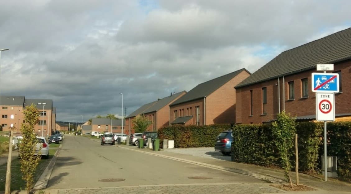 En tant qu'expert en code de la route belge, je vais analyser l'image de cette question d'examen de conduite et vous fournir une description détaillée.
Description détaillée de la situation routière :
Type de situation routière :
L'image présente une vue de face d'une rue résidentielle, probablement située dans un nouveau lotissement ou un quartier récemment développé, étant donné l'aspect moderne et uniforme des habitations. Il s'agit d'une route d'accès local, bordée de maisons, avec des véhicules stationnés. La chaussée s'éloigne en une légère courbe vers la gauche au loin.
Éléments visibles :
Chaussée : La route est asphaltée, de couleur grise, et semble suffisamment large pour permettre le croisement de deux véhicules, bien que la présence de voitures stationnées puisse en réduire la largeur effective. Des regards d'égout sont visibles sur la chaussée. Sur la gauche, le bord de la route présente des pavés et une petite bande de verdure.
Habitations : De part et d'autre de la rue, on observe des maisons mitoyennes ou semi-mitoyennes de style contemporain, construites en briques rouges et dotées de toits en tuiles sombres. Elles possèdent plusieurs étages et des fenêtres régulières.
Végétation : Des haies bien entretenues délimitent les propriétés sur la droite. De jeunes arbres sont plantés le long de la chaussée, notamment sur la gauche.
Véhicules : Plusieurs véhicules sont stationnés le long de la rue :
Sur la gauche, au moins trois voitures (un SUV/hatchback blanc, une berline sombre, et une autre voiture sombre) sont garées en épi ou en parallèle par rapport au sens de la prise de vue.
Sur la droite, un SUV de couleur foncée (bleu marine ou noir) est garé sur une allée privée devant une maison, derrière la haie.
Équipements urbains : Des poteaux d'éclairage public sont visibles des deux côtés de la rue, s'étendant vers l'horizon. Des poubelles vertes sont alignées le long de la haie sur la droite.
Ciel : Le ciel est couvert, avec des nuages gris, suggérant une journée nuageuse ou une fin de journée.
Position et direction des véhicules :
L'image est prise du point de vue d'un conducteur se dirigeant dans la rue. Tous les véhicules visibles sont stationnés et ne sont donc pas en mouvement. Les voitures sur la gauche sont garées globalement dans le sens de la progression de la caméra. Le SUV sur la droite est stationné dans une propriété privée.
Panneaux de signalisation présents et leur signification :
Sur le côté droit de la route, juste après le point de prise de vue de la caméra, un poteau de signalisation porte deux panneaux importants, superposés :
Panneau supérieur (le plus petit, blanc, rectangulaire) : Il s'agit d'une petite plaque additionnelle blanche dont le texte est difficilement lisible, mais qui semble indiquer le nom du quartier ou de la zone, potentiellement précédé de "FIN" (ex: "FIN COLLEN").
Panneau intermédiaire (bleu, rectangulaire, avec barre rouge diagonale) : C'est un panneau de type F12b ou F12d (Fin de zone résidentielle ou Fin de zone de rencontre). Il représente une voiture et une silhouette de personne (souvent un enfant jouant), barrées d'une ligne diagonale rouge. Ce panneau indique que l'on sort d'une zone où des règles spécifiques s'appliquaient (telles que la priorité aux piétons, le stationnement organisé, etc.).
• Panneau inférieur (blanc, carré, avec un cercle rouge et "30") : C'est un panneau de type F4a (Début de zone 30). Il indique l'entrée dans une zone où la vitesse maximale autorisée pour tous les usagers de la route est de 30 kilomètres par heure. Cette limitation s'applique à l'ensemble de la zone jusqu'à ce qu'un panneau de fin de zone 30 (F4b) soit rencontré.
Tout autre élément pertinent pour comprendre la situation :
La disposition des panneaux est cruciale : on quitte une zone spécifique (résidentielle/de rencontre) pour entrer immédiatement dans une "Zone 30". La question "En deçà de ces signaux routiers, la vitesse maximale autorisée est de :" signifie "Après avoir passé ces signaux, dans la direction de la marche, quelle est la vitesse maximale autorisée ?". Par conséquent, le panneau F4a ("ZONE 30") est celui qui définit la nouvelle limitation de vitesse pour le tronçon de route que l'on s'apprête à emprunter. La combinaison de ces panneaux est courante en Belgique pour marquer la transition entre différents régimes de circulation dans les zones urbaines ou résidentielles.