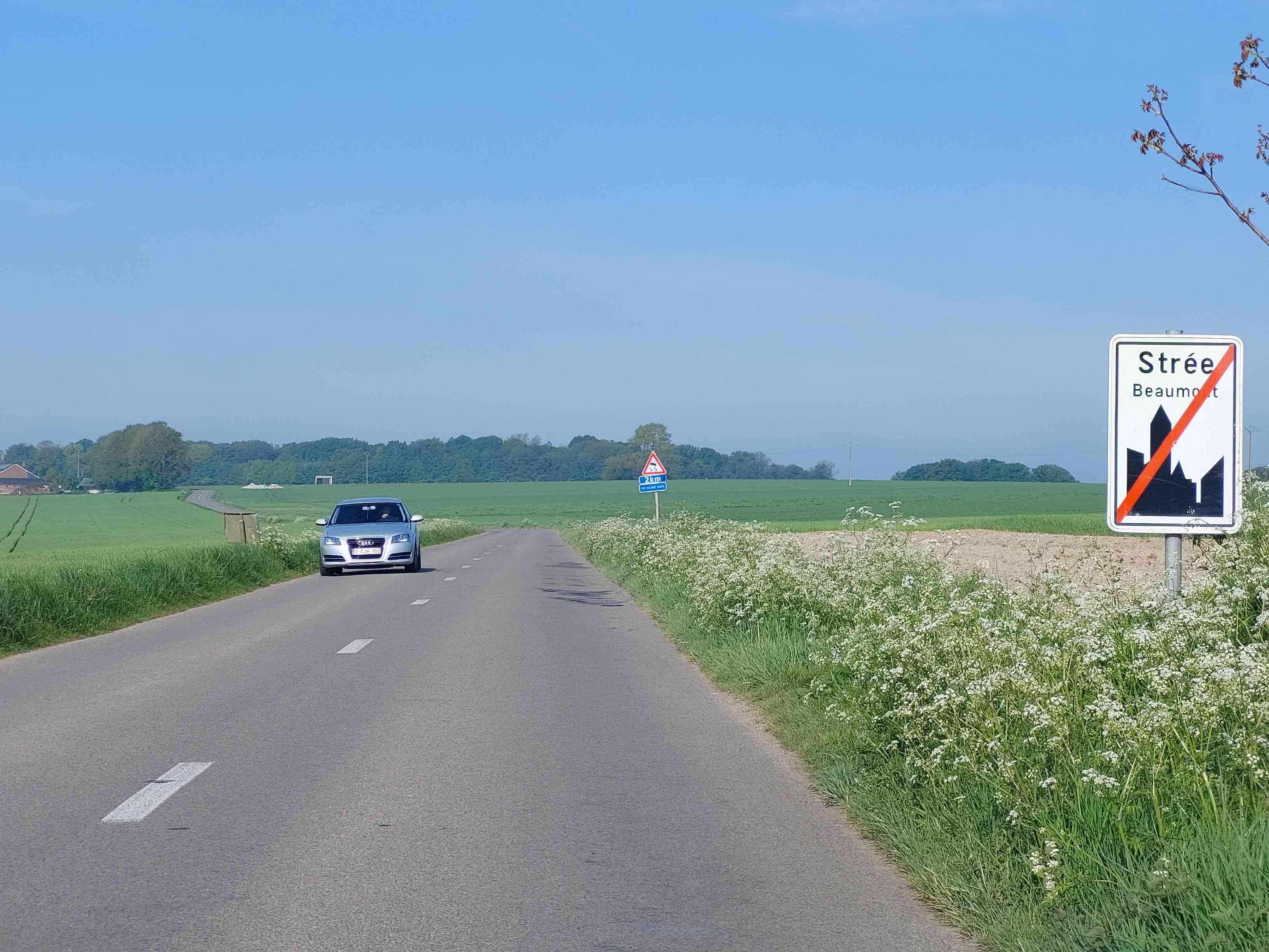 Absolument ! Voici une description détaillée de la situation routière présente dans l'image, en tant qu'expert du code de la route belge :
Description Détaillée de la Situation Routière (Contexte Wallon)
La scène se déroule sur une route de campagne ouverte, sous un ciel bleu clair, suggérant une journée ensoleillée et une bonne visibilité. L'image est prise du point de vue d'un conducteur avançant sur cette route.
Type de situation routière :
Il s'agit d'une section de route bidirectionnelle, à chaussée unique, en milieu rural ou semi-rural. La route est bordée de champs de cultures verdoyants et d'accotements herbeux fleuris (fleurs blanches de type ombellifères). Le paysage est dégagé et ouvert, avec une ligne d'arbres à l'horizon en arrière-plan. Il n'y a pas d'intersection immédiate visible au premier plan. La situation principale est celle de la sortie d'une agglomération.
Éléments visibles :
La Chaussée : Asphalte gris foncé, en bon état apparent.
Marquages au sol : Au centre de la chaussée, une ligne discontinue blanche (segments) est visible, séparant les deux sens de circulation et autorisant le dépassement. Sur le côté droit, une ligne blanche continue marque le bord de la chaussée.
Accotements : Larges accotements couverts d'herbe haute et de petites fleurs blanches.
Paysage : Vastes champs verts de part et d'autre de la route, délimitant le caractère rural du lieu. Une bande d'arbres dense forme l'horizon lointain. Quelques bâtiments de ferme ou annexes sont à peine visibles à gauche dans le lointain.
Véhicules : Une voiture de couleur gris argenté (une Audi A3, reconnaissable à sa calandre) approche en sens inverse du point de vue de la caméra. Elle roule sur sa voie de droite. Sa plaque d'immatriculation belge est "1-CJH-102".
Infrastructure : Un poteau électrique est visible à gauche de la chaussée, plus loin sur la route. Des branches d'arbre feuillues sont visibles dans le coin supérieur droit de l'image, partiellement floues.
Position et direction des véhicules :
Le conducteur, d'après la question, est en train d'aborder la zone signalisée. La voiture visible (Audi) est en sens inverse, circulant vers l'observateur. Pour le conducteur qui pose la question, cela signifie qu'il est sur le point de sortir de l'agglomération de Strée. L'Audi vient donc de la direction que le conducteur de la question s'apprête à emprunter (c'est-à-dire, venant de l'extérieur de l'agglomération, mais elle est déjà passée le panneau de fin d'agglomération dans son sens de circulation).
Panneaux de signalisation présents et leur signification :
Sur la droite, au premier plan (panneau principal pour la question) :
Un panneau de signalisation de type F1a (Fin d'agglomération). C'est un panneau rectangulaire blanc à bordure noire. Il porte le nom de la localité "Strée" en caractères plus grands, et en dessous "Beaumont" en caractères plus petits (Beaumont étant la commune à laquelle Strée est rattachée). Le panneau comporte une silhouette stylisée de bâtiments de ville, barrée d'une épaisse ligne diagonale rouge.
Signification : Ce panneau indique la fin de l'agglomération de Strée. À partir de ce point, les règles de circulation spécifiques aux agglomérations (dont la limitation de vitesse à 50 km/h) ne sont plus d'application.
Sur la gauche, plus loin sur la route (avant l'Audi) :
Un panneau de danger de type A13 (Chaussée glissante). C'est un panneau triangulaire à bord rouge et fond blanc, avec un pictogramme noir représentant une voiture qui dérape.
En dessous, un panneau additionnel rectangulaire bleu porte l'inscription "2km" et "SUR CHAUSSÉE HUMIDE".
Signification : Ce panneau avertit les usagers de la route que, sur les 2 kilomètres suivants, la chaussée peut être particulièrement glissante, notamment lorsque celle-ci est humide. Il s'agit d'un avertissement de danger qui n'impose pas une limitation de vitesse fixe mais qui appelle à la prudence et à une adaptation de la vitesse et de la conduite aux conditions. Ce panneau est placé avant le panneau de fin d'agglomération pour le conducteur venant de l'autre sens, ou après pour le conducteur sortant de l'agglomération et le laissant derrière lui.
Tout autre élément pertinent pour comprendre la situation :
La question spécifie "Je suis en région Wallonne". Ceci est une information cruciale. En Belgique, la vitesse maximale autorisée hors agglomération varie selon les régions :
En Région Wallonne, la vitesse maximale autorisée sur les routes ordinaires hors agglomération est de 90 km/h, sauf indication contraire par un panneau spécifique.
• En Région Flamande et en Région de Bruxelles-Capitale, cette vitesse est de 70 km/h.
En synthèse pour la question : Le signal routier auquel la question fait référence ("ce signal routier") est le panneau de fin d'agglomération (F1a) "Strée Beaumont" sur la droite. La question est "En deçà de ce signal routier, ma vitesse est limitée à :". "En deçà" signifie "au-delà de" ou "une fois passé ce signal". Donc, une fois que le conducteur a dépassé ce panneau de fin d'agglomération et se trouve dans la zone rurale, en Wallonie, la limitation de vitesse générale s'applique.