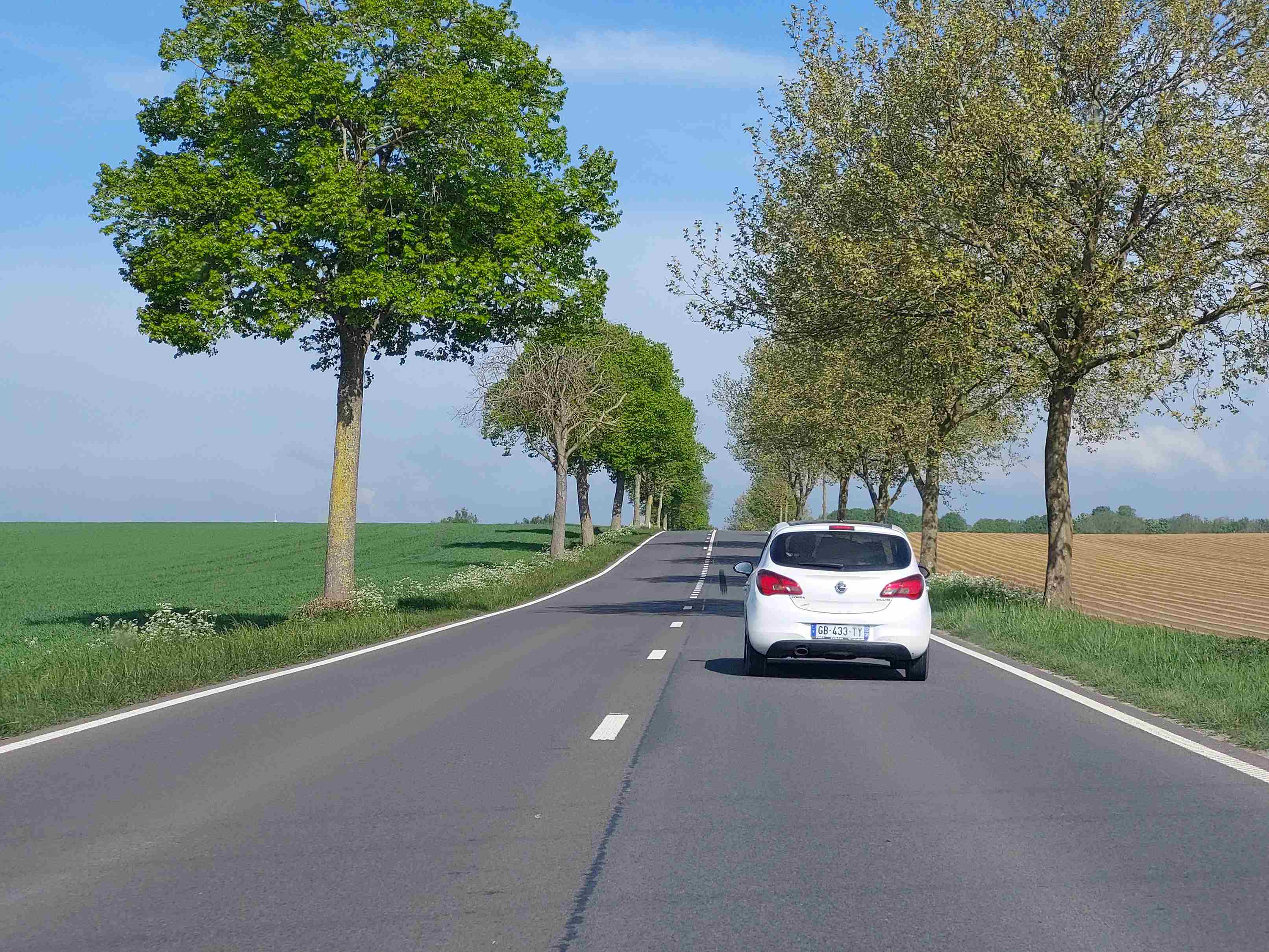 En tant qu'expert du code de la route belge, voici la description détaillée de la situation routière présentée sur l'image, pour comprendre le contexte de la question : "Je circule plus vite que ce véhicule, puis-je le dépasser maintenant ?"
Description détaillée de la situation routière
Type de situation routière:
La scène se déroule sur une route bidirectionnelle à chaussée unique, en milieu rural ou semi-rural. Il s'agit d'une route nationale ou provinciale, bordée des deux côtés par une succession d'arbres formant une sorte d'allée ou d'avenue arborée. La route présente une légère montée ou un sommet de côte (crête) à l'horizon, ce qui est un élément crucial pour l'évaluation de la sécurité du dépassement. Il n'y a pas d'intersection, de carrefour, de passage à niveau ou de passage pour piétons visible à proximité immédiate.
Éléments visibles:
Véhicules:
Une voiture de couleur blanche, de type berline compacte (ressemblant à une Opel Corsa), est visible devant le véhicule du conducteur (qui prend la photo). Elle circule dans la même direction.
Aucun autre véhicule n'est visible, que ce soit en sens inverse ou stationné.
Panneaux de signalisation:
Aucun panneau de signalisation vertical n'est visible sur l'image, ni sur les côtés de la route ni au-dessus.
Feux de circulation:
Aucun feu de circulation n'est présent.
Marquages au sol:
Ligne centrale: La chaussée est dotée d'une ligne discontinue blanche axiale, indiquant qu'elle sépare deux sens de circulation et que le dépassement est généralement autorisé si les conditions le permettent.
Lignes latérales: Des lignes continues blanches délimitent les bords de la chaussée (accotements ou bas-côtés).
Environnement:
Le ciel est clair, majoritairement bleu avec quelques fines traces de nuages, suggérant une bonne visibilité météorologique.
Des arbres feuillus bordent la route des deux côtés, avec une végétation basse sur les accotements.
De vastes champs agricoles s'étendent de chaque côté de la route : un champ vert à gauche (cultures en croissance) et un champ labouré de couleur marron-ocre à droite.
Position et direction des véhicules:
Le conducteur (dont la perspective est celle de la photo) se trouve derrière la voiture blanche.
La voiture blanche roule devant le conducteur, positionnée au centre de sa voie de circulation.
Les deux véhicules circulent dans la même direction, sur la partie droite de la chaussée.
Panneaux de signalisation présents et leur signification:
Aucun panneau de signalisation vertical n'est présent sur l'image. Cela signifie qu'il n'y a pas de limitation de vitesse spécifique imposée par un panneau, ni d'interdiction de dépasser signalée par un panneau B1, ni d'indication de priorité ou de danger immédiat autre que ce qui est intrinsèque à la configuration de la route.
Tout autre élément pertinent pour comprendre la situation:
Visibilité latérale: La visibilité est bonne sur les côtés (champs ouverts).
Visibilité vers l'avant: C'est l'élément le plus important. La route monte légèrement et se dirige vers un sommet de côte (crête). La visibilité au-delà de ce sommet est limitée voire nulle. On ne peut pas savoir ce qui arrive en sens inverse ou ce qui se passe juste derrière le sommet, ce qui est un facteur déterminant pour la sécurité d'un dépassement. L'absence de visibilité suffisante au-delà du sommet de la côte est une condition d'interdiction de dépassement selon le code de la route belge.
Contexte pour la question: La question porte sur la légalité et la sécurité d'un dépassement. Bien que la ligne discontinue centrale autorise en principe le dépassement, la présence d'un sommet de côte qui limite la visibilité vers l'avant est une condition spécifique où le dépassement est généralement interdit en Belgique pour des raisons de sécurité.