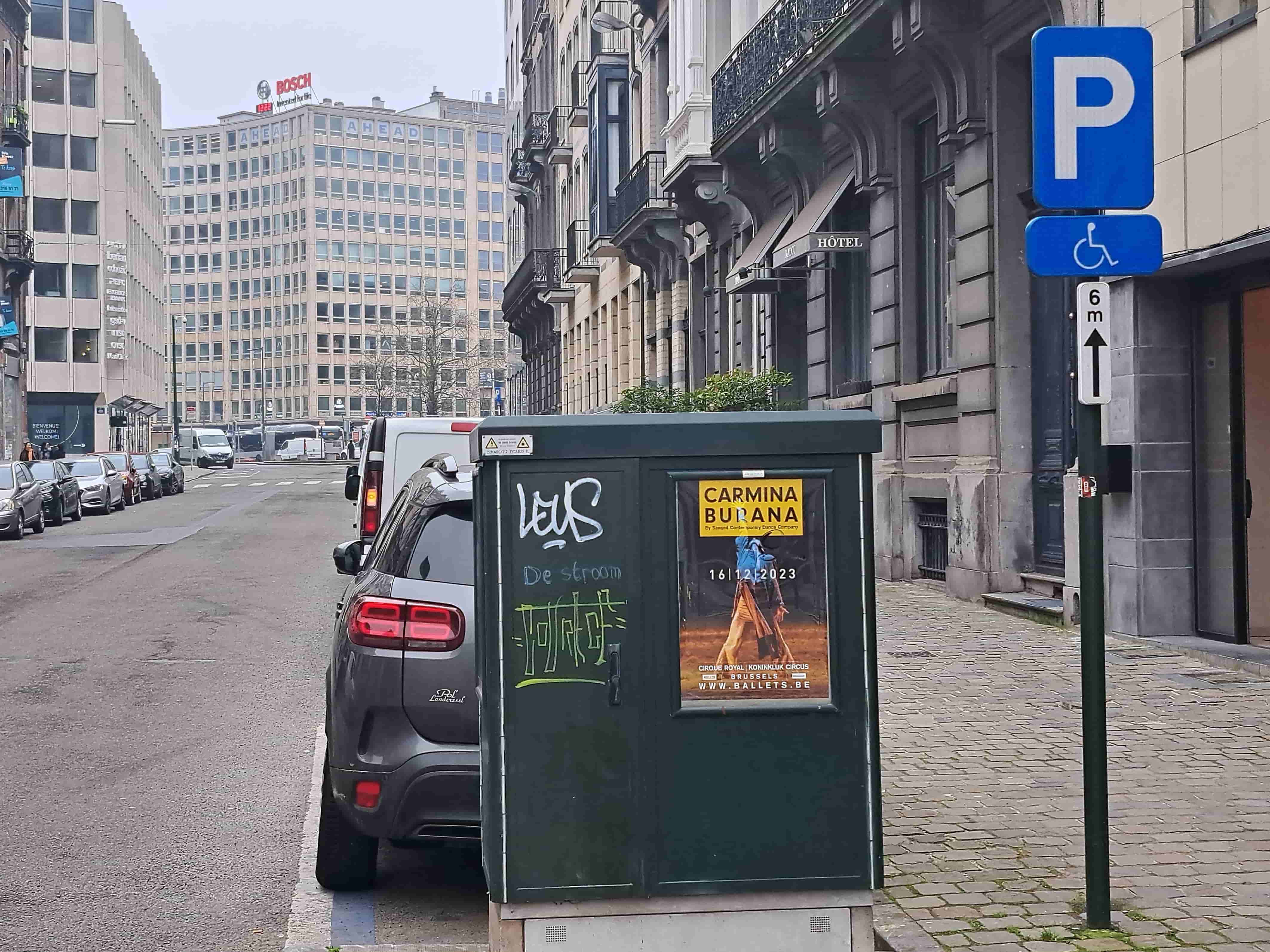 Voici une description détaillée de la situation routière présentée dans l'image, en tant qu'expert en code de la route belge :
Description Détaillée de la Situation Routière
Type de situation routière :
L'image représente une scène de rue urbaine animée, typique d'un centre-ville ou d'un quartier d'affaires. Il s'agit d'une voie de circulation asphaltée à sens unique ou bidirectionnelle (bien que le sens ne soit pas explicitement déterminé, les voitures garées le long du trottoir droit suggèrent un alignement), bordée par un trottoir large à droite et des bâtiments. On observe une ligne de véhicules garés en stationnement parallèle sur le côté droit de la chaussée. Plus loin, on distingue un passage pour piétons (bandes zébrées blanches).
Éléments visibles :
Véhicules : Une file de voitures est stationnée le long du trottoir droit. Au premier plan, l'arrière d'un SUV gris foncé (ressemblant à un Citroën C5 Aircross) est visible, suivi d'une camionnette blanche. Plus loin, d'autres voitures sont garées. Sur la chaussée, au loin, on aperçoit une autre camionnette blanche et au moins deux bus.
Panneaux de signalisation : Un poteau vert supporte une pile de panneaux de signalisation très pertinents pour la question :
Panneau principal : Un grand panneau carré à fond bleu avec un "P" blanc au centre (Panneau E9a), indiquant un emplacement de stationnement.
Premier panneau additionnel : Un panneau rectangulaire à fond bleu avec un pictogramme blanc d'une personne en fauteuil roulant (Panneau M4), indiquant que le stationnement est réservé aux personnes handicapées.
Deuxième panneau additionnel : Un panneau rectangulaire blanc vertical avec l'inscription "6 m" et une flèche noire pointant vers le haut, indiquant que la prescription s'applique "au-delà du signal" (dans le sens de la marche) sur une distance de 6 mètres.
Feux : Aucun feu de signalisation ou feu tricolore n'est visible dans le champ immédiat. Les feux arrière du SUV et de la camionnette sont visibles mais éteints.
Marquages au sol :
Une fine ligne bleue est visible sur le trottoir/bordure, juste devant le pneu avant du SUV, à l'endroit où le caisson technique commence. Cette ligne pourrait délimiter une zone de stationnement spécifique ou un emplacement réservé.
Plus loin sur la chaussée, on distingue un passage pour piétons (zébra).
Autres éléments :
Caisson technique : Un grand caisson vert foncé, probablement électrique ou de télécommunications, est placé sur le trottoir, juste après le poteau des panneaux et devant le SUV. Il est orné de graffitis et d'une affiche publicitaire pour "Carmina Burana".
Bâtiments : Sur la droite, des bâtiments anciens à plusieurs étages avec des balcons et des façades classiques, dont un porte une enseigne "HÔTEL". Sur la gauche et en arrière-plan, des immeubles de bureaux modernes, dont un grand bâtiment clairement identifié par les logos "BOSCH" et "AHEAD".
Trottoir : Pavé de dalles ou de pavés sur le côté droit de la rue.
Éclairage : Le ciel est couvert, la lumière est diffuse.
Position et direction des véhicules :
Les véhicules garés sur le côté droit de la rue sont tous stationnés parallèlement au trottoir, orientés dans la même direction, s'éloignant du point de vue de l'observateur. La camionnette blanche et les bus lointains semblent également se déplacer ou être orientés dans la même direction que les voitures garées.
Panneaux de signalisation présents et leur signification (en relation avec le Code de la route belge) :
Panneau E9a (Stationnement) : Indique un emplacement où le stationnement est autorisé.
Panneau additionnel M4 (Personne handicapée) : Ce panneau modifie le panneau E9a en indiquant que le stationnement sur cet emplacement est réservé exclusivement aux véhicules utilisés par les personnes handicapées titulaires d'une carte spéciale de stationnement.
• Panneau additionnel "6 m" avec flèche vers le haut : Ce panneau précise la portée de la prescription. La flèche vers le haut (symbole M10) signifie que la réservation du stationnement pour personnes handicapées s'applique après le panneau, dans le sens de la marche, et ce, sur une distance de 6 mètres à partir de la position du panneau.
Tout autre élément pertinent :
La question d'examen mentionne explicitement "Au-delà du signal routier, sur une distance de 6 mètres". Cela correspond parfaitement à l'interprétation des panneaux de signalisation. Le SUV garé juste après le caisson technique (lui-même après le panneau) se trouve potentiellement dans cette zone de 6 mètres. La ligne bleue au sol près du véhicule peut confirmer une délimitation de place, mais la portée des 6 mètres du panneau prime. La présence d'un hôtel peut parfois entraîner des zones de "dépose-minute" ou de courtes haltes, mais les panneaux de signalisation ici définissent clairement la règle de stationnement prévalente.
En résumé, la situation montre un emplacement de stationnement réservé aux personnes handicapées, dont la validité commence au niveau du panneau et s'étend sur une distance de 6 mètres dans le sens de la marche.