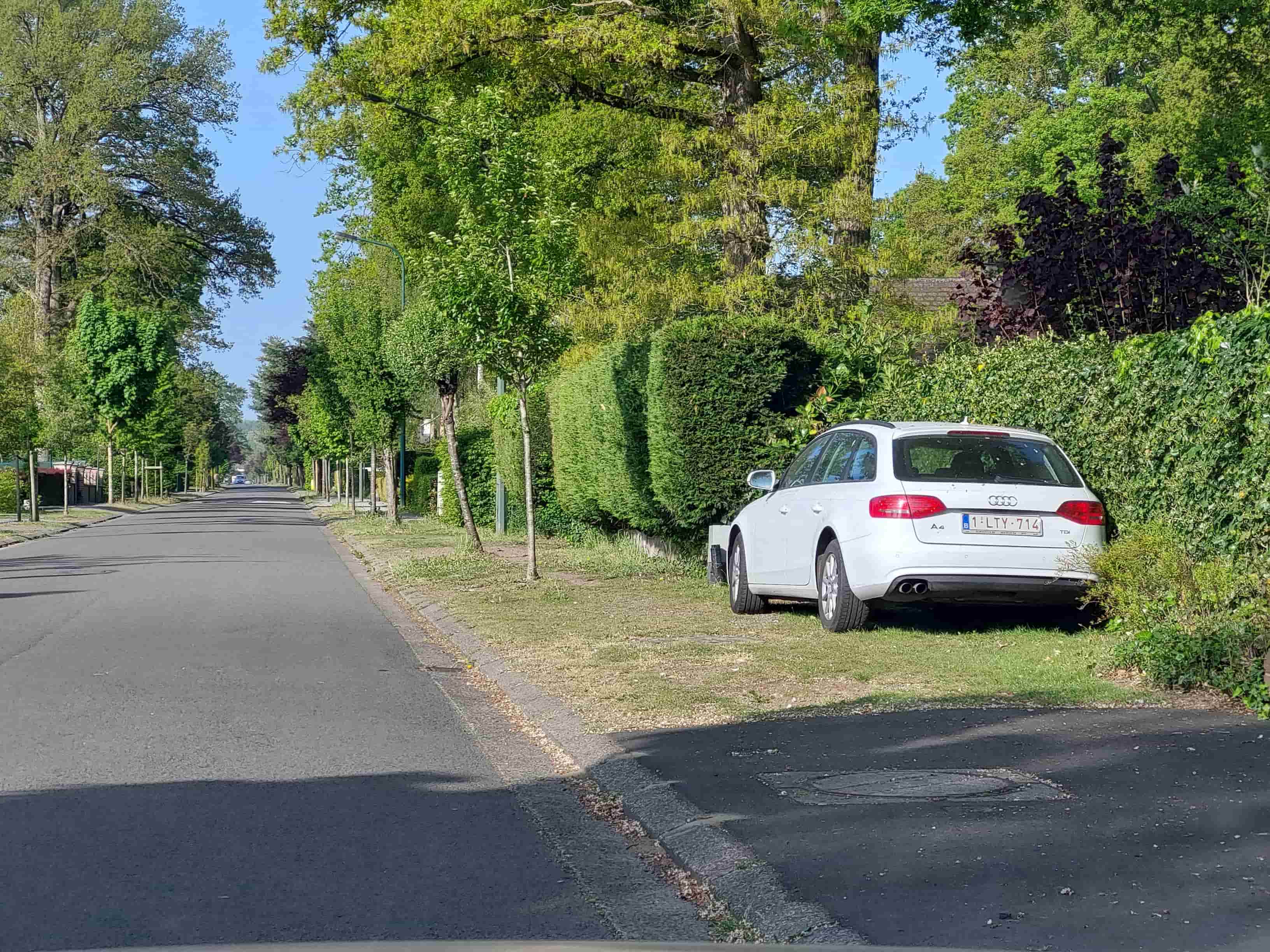 En tant qu'expert en code de la route belge, voici une description détaillée de la situation routière présentée sur l'image, en relation avec la question posée :
Contexte de la question : "Puis-je arrêter ma voiture blanche sur cet accotement en agglomération ?"
Description détaillée de la situation routière :
Type de situation routière :
L'image présente une portion de route publique, vraisemblablement une rue résidentielle ou de desserte locale, traversant ce qui est implicitement décrit par la question comme une agglomération (zone bâtie).
La chaussée est bidirectionnelle (bien que l'absence de marquage central ou de sens unique ne le confirme pas absolument, c'est l'hypothèse par défaut pour ce type de rue).
Il n'y a pas d'intersection, de carrefour, de passage piéton, ni de signalisation lumineuse (feux de circulation) visible dans l'immédiat. La route s'étend en ligne droite vers l'horizon.
Éléments visibles :
Chaussée : La surface est en asphalte, de couleur grise foncée, et semble en bon état. Elle est d'une largeur suffisante pour permettre la circulation dans les deux sens ou le croisement de véhicules.
Bordure de trottoir / Accotement : Un trottoir surélevé, matérialisé par une bordure en béton gris clair, sépare clairement la chaussée de l'accotement enherbé sur la droite.
Accotement enherbé (à droite) : C'est une bande de terre recouverte d'herbe et de quelques herbes sèches, située immédiatement après la bordure de trottoir. C'est sur cet accotement que le véhicule blanc est stationné.
Véhicule : Une voiture blanche, de marque Audi A4 Avant (break), immatriculée en Belgique (plaque "1-LTY-714").
Végétation :
Sur le côté droit, derrière la voiture, se trouve une haie dense et taillée (probable clôture de propriété), ainsi que d'autres arbustes et buissons. Des arbres feuillus plus grands sont visibles en arrière-plan.
Sur le côté gauche de la chaussée, une rangée d'arbres plus jeunes et des arbustes sont plantés le long de la route, séparés de la chaussée par une étroite bande d'herbe et une bordure similaire à celle de droite.
Éléments d'infrastructure : Des lampadaires (poteaux métalliques vert foncé) sont visibles le long du côté gauche de la route, suggérant un éclairage public. Un regard ou une plaque d'égout est visible sur la chaussée au premier plan.
Environnement général : L'ambiance est celle d'une journée ensoleillée, avec un ciel bleu dégagé. Les ombres projetées sont nettes.
Position et direction des véhicules :
La voiture blanche est stationnée parallèlement à la chaussée et à la bordure de trottoir, mais avec ses quatre roues entièrement sur l'accotement enherbé, à droite de la voie de circulation.
L'arrière du véhicule est légèrement tourné vers l'observateur/la caméra. La voiture fait face à la direction générale de la route, s'éloignant de la caméra.
Il n'y a pas d'autres véhicules visibles en mouvement ou stationnés sur la chaussée.
Panneaux de signalisation présents et leur signification :
Aucun panneau de signalisation routière spécifique (comme interdiction de stationnement, limitation de vitesse, etc.) n'est visible dans le champ de l'image.
Cependant, le contexte de la question ("en agglomération") est crucial. En Belgique, l'entrée en agglomération est généralement signalée par le panneau F1 et la sortie par le panneau F3. L'absence de ces panneaux dans l'image ne signifie pas que nous sommes hors agglomération, mais que le point de vue se situe à l'intérieur de celle-ci, comme le stipule la question. Les règles générales du code de la route pour les agglomérations s'appliquent donc.
Tout autre élément pertinent :
L'accotement enherbé n'est pas un trottoir aménagé pour les piétons de manière claire (pas de dalles ou de revêtement uniforme), mais plutôt une bande verte servant de séparation entre la propriété et la chaussée, délimitée par une bordure.
• La présence de la bordure et de l'herbe indique une distinction claire entre la chaussée (réservée à la circulation) et l'espace latéral.
Cette description fournit tous les éléments visuels et contextuels nécessaires pour analyser la légalité de l'arrêt ou du stationnement du véhicule blanc selon le code de la route belge en agglomération.