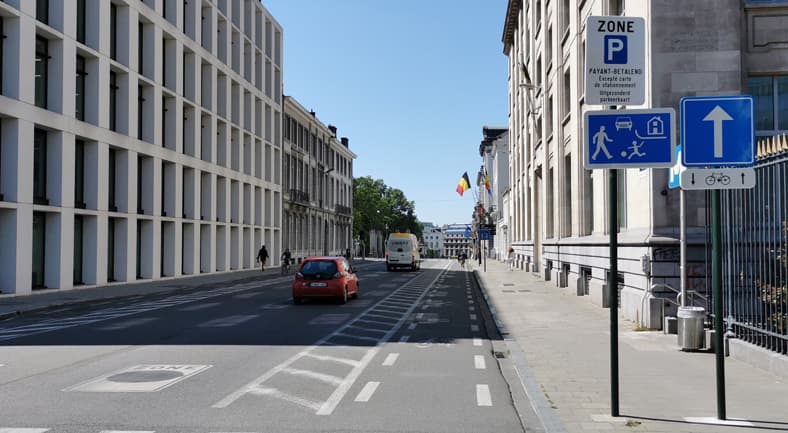 Voici une description détaillée de l'image, en tant qu'expert du code de la route belge, pour comprendre le contexte de la question "Au-delà de ces signaux routiers :".
Description Détaillée de la Situation Routière (Belgique)
L'image présente une scène de rue urbaine en Belgique, par une journée ensoleillée et dégagée. Elle est prise depuis le milieu de la chaussée, regardant vers l'avant dans le sens de la circulation.
Type de situation routière :
Il s'agit d'une artère urbaine, probablement une rue à sens unique (fortement suggéré par la signalisation et la direction des véhicules), bordée de bâtiments. La signalisation verticale et horizontale indique l'entrée dans une zone spécifique soumise à une réglementation particulière, notamment une zone de stationnement payant et, surtout, une "zone de rencontre".
Éléments visibles :
Véhicules :
Une petite voiture rouge (type citadine) est visible sur la chaussée, s'éloignant de l'observateur.
Un fourgon blanc de type utilitaire (avec l'inscription "COOXY" à l'arrière) la précède, également s'éloignant.
Un cycliste est discernable plus loin sur la gauche de la chaussée.
Des piétons sont visibles sur les trottoirs, loin devant sur la gauche et la droite.
Marquages au sol :
Au premier plan, un large marquage au sol rectangulaire blanc avec l'inscription "ZONE" est clairement visible sur l'asphalte. Ce marquage renforce la signalisation verticale d'une zone réglementée.
Une ligne blanche continue délimite le trottoir de la chaussée sur la droite.
Sur la gauche de la chaussée, une bande délimitée par des lignes pointillées blanches est présente, suggérant un aménagement pour cyclistes ou un espace partagé.
Une ligne blanche continue sépare cette bande de la voie principale de circulation.
Infrastructures et bâtiments :
Sur la gauche, on observe un grand bâtiment moderne aux lignes épurées.
Sur la droite, des bâtiments plus classiques, avec des fenêtres régulières et une architecture plus ancienne, bordent la rue.
Des drapeaux belges sont visibles plus loin sur la droite, flottant au-dessus des bâtiments.
Du mobilier urbain (poubelle, poteaux de signalisation, lampadaires) est également présent.
Position et direction des véhicules :
La voiture rouge et le fourgon blanc circulent sur la voie de droite (la voie principale) et s'éloignent de l'observateur. Le cycliste est également en mouvement dans la même direction, sur l'aménagement cyclable à gauche. L'ensemble des véhicules visibles se dirige dans le même sens, ce qui est cohérent avec une rue à sens unique.
Panneaux de signalisation présents et leur signification (tous situés sur le trottoir de droite, à proximité de l'observateur) :
Panneau supérieur (Signal E9a - Zone de stationnement payant) :
Il s'agit d'un panneau rectangulaire vertical bleu et blanc, avec un "P" blanc sur fond bleu et le texte "ZONE PAYANT-BETALEND" en dessous.
Il est complété par un panonceau indiquant "Excepté carte de stationnement - Uitgezonderd parkeerkaart".
Signification : Ce panneau indique l'entrée dans une zone de stationnement où le stationnement est payant, à l'exception des véhicules munis d'une carte de stationnement spécifique (par exemple, riverain, personne à mobilité réduite).
Panneau central (Signal F12b - Début d'une zone de rencontre / zone résidentielle) :
Un panneau rectangulaire horizontal bleu, représentant stylisés une voiture, un piéton, un enfant jouant au ballon et une maison.
Signification : Ce signal marque le début d'une zone de rencontre. Les règles spécifiques à cette zone sont les suivantes :
La vitesse est limitée à 20 km/h.
Les piétons peuvent utiliser toute la largeur de la voie publique et ont la priorité sur les véhicules.
Les véhicules ne peuvent pas gêner les piétons.
Le stationnement est uniquement autorisé aux endroits désignés par une signalisation spécifique (souvent des marquages au sol ou d'autres panneaux).
Panneau inférieur (Signal D1 - Obligation d'aller tout droit avec panonceau) :
Un panneau carré bleu avec une flèche blanche pointant vers le haut (Signal D1).
Signification principale : Ce panneau impose l'obligation d'aller tout droit pour tous les usagers de la route concernés par cette voie.
Panonceau : Un panonceau blanc sur fond bleu est situé sous la flèche, représentant un vélo avec une flèche vers le haut et une flèche vers le bas, séparées par une ligne horizontale.
• Signification du panonceau : Ce panonceau indique que l'obligation d'aller tout droit s'applique également aux cyclistes, ou qu'il existe un aménagement cyclable spécifique (potentiellement bidirectionnel, ou une direction particulière pour les vélos) dans cette direction.
Autres éléments pertinents pour comprendre la situation :
La combinaison du marquage au sol "ZONE" et du panneau F12b (Zone de rencontre) est redondante mais insiste sur l'importance de cette réglementation. La présence d'une bande cyclable et le panonceau "vélo" sous la flèche D1 soulignent l'intégration des cyclistes dans cet environnement urbain et la nécessité de partager l'espace de manière sécuritaire, surtout dans une zone de rencontre où les usagers faibles ont une priorité accrue. La situation appelle à une vigilance particulière en raison des règles spécifiques de la zone de rencontre et de la coexistence de différents types d'usagers.