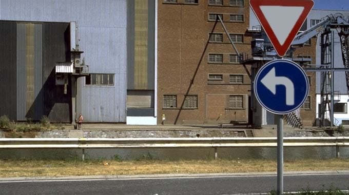 Absolument ! En tant qu'expert du code de la route belge, voici une analyse détaillée de cette image de question d'examen :
Description Détaillée de la Situation Routière (Contexte : "A ce carrefour,")
Cette image présente une situation routière urbaine ou industrielle, visible depuis la perspective d'un conducteur se déplaçant sur une route asphaltée.
Type de situation routière :
Nous sommes sur une route asphaltée, visible en premier plan.
L'intitulé de la question ("A ce carrefour,") indique que le conducteur est en train d'approcher ou se trouve déjà à une intersection. L'intersection elle-même n'est pas entièrement visible dans le cadre de l'image, mais les panneaux de signalisation confirment sa proximité immédiate et la nécessité de préparer une manœuvre.
Sur le côté droit de la route, on observe un accotement (ou une bande de terre) enherbée et sèche, suivi d'une glissière de sécurité métallique (garde-fou blanc) qui sépare la route d'un niveau inférieur ou d'un autre aménagement, probablement un quai ou une zone industrielle.
Éléments visibles :
Route : Une portion de route goudronnée, dont la largeur de la voie de circulation n'est pas clairement définie par des marquages au sol visibles dans le champ.
Végétation : De l'herbe sèche le long de l'accotement, et quelques petits arbustes ou buissons au-delà de la glissière de sécurité.
Structures industrielles : En arrière-plan, plusieurs bâtiments massifs et de style industriel sont visibles. On distingue un grand bâtiment en tôle ondulée (gris/bleu avec des bandes verticales jaunes), et un bâtiment en briques rouges. Des éléments de structures métalliques, ressemblant à des grues ou des supports, sont également présents, renforçant l'impression d'un environnement portuaire ou d'une zone d'activités industrielles.
Personne : Une personne est visible à pied dans le lointain, entre les bâtiments et la glissière de sécurité, sur ce qui semble être un quai ou un trottoir élevé, bien à l'écart de la voie de circulation principale.
Véhicules : Aucun véhicule n'est visible sur la route au premier plan, ni dans l'axe du regard du conducteur.
Feux de signalisation : Aucun feu tricolore n'est présent.
Marquages au sol : Aucun marquage au sol (lignes continues, discontinues, flèches directionnelles, lignes d'arrêt) n'est visible sur la chaussée dans le champ proche de l'image.
Position et direction des véhicules s'il y en a :
Comme mentionné, aucun véhicule n'est visible sur la chaussée principale, impliquant que le conducteur de la voiture caméra est le seul usager dans ce champ visuel immédiat.
Panneaux de signalisation présents et leur signification (Code de la Route Belge) :
Panneau supérieur : Il s'agit d'un panneau B1 (triangle inversé, bord rouge, fond blanc).
Signification : "CÉDEZ LE PASSAGE". Ce panneau impose au conducteur de céder le passage à tout véhicule venant d'une autre direction au carrefour qui s'annonce. Le conducteur doit, si nécessaire, ralentir ou même s'arrêter pour laisser passer les autres usagers en toute sécurité.
Panneau inférieur : Il s'agit d'un panneau D1 (rond bleu, flèche blanche tournant à gauche).
Signification : "DIRECTION OBLIGATOIRE À LA PROCHAINE INTERSECTION : À GAUCHE". Ce panneau indique au conducteur qu'il doit impérativement tourner à gauche à l'intersection qui suit immédiatement ce panneau. Il n'est pas autorisé à continuer tout droit ni à tourner à droite.
Tout autre élément pertinent :
L'association des panneaux B1 et D1 est cruciale : Le conducteur doit non seulement céder le passage à l'intersection (B1), mais il est également obligé de prendre la direction à gauche (D1). Cela signifie qu'il devra s'assurer que la voie est libre pour s'engager en toute sécurité vers la gauche.
L'absence de marquages au sol sur la chaussée principale place toute l'importance sur les panneaux pour la régulation du trafic à l'approche de cette intersection.
L'environnement industriel pourrait impliquer une circulation de véhicules lourds, une visibilité potentiellement réduite à l'intersection à cause des bâtiments, ou la présence inattendue d'engins industriels.
En résumé, la situation décrit l'approche d'un carrefour où le conducteur doit impérativement céder le passage et tourner à gauche, guidé par une signalisation verticale claire, dans un environnement aux allures industrielles.