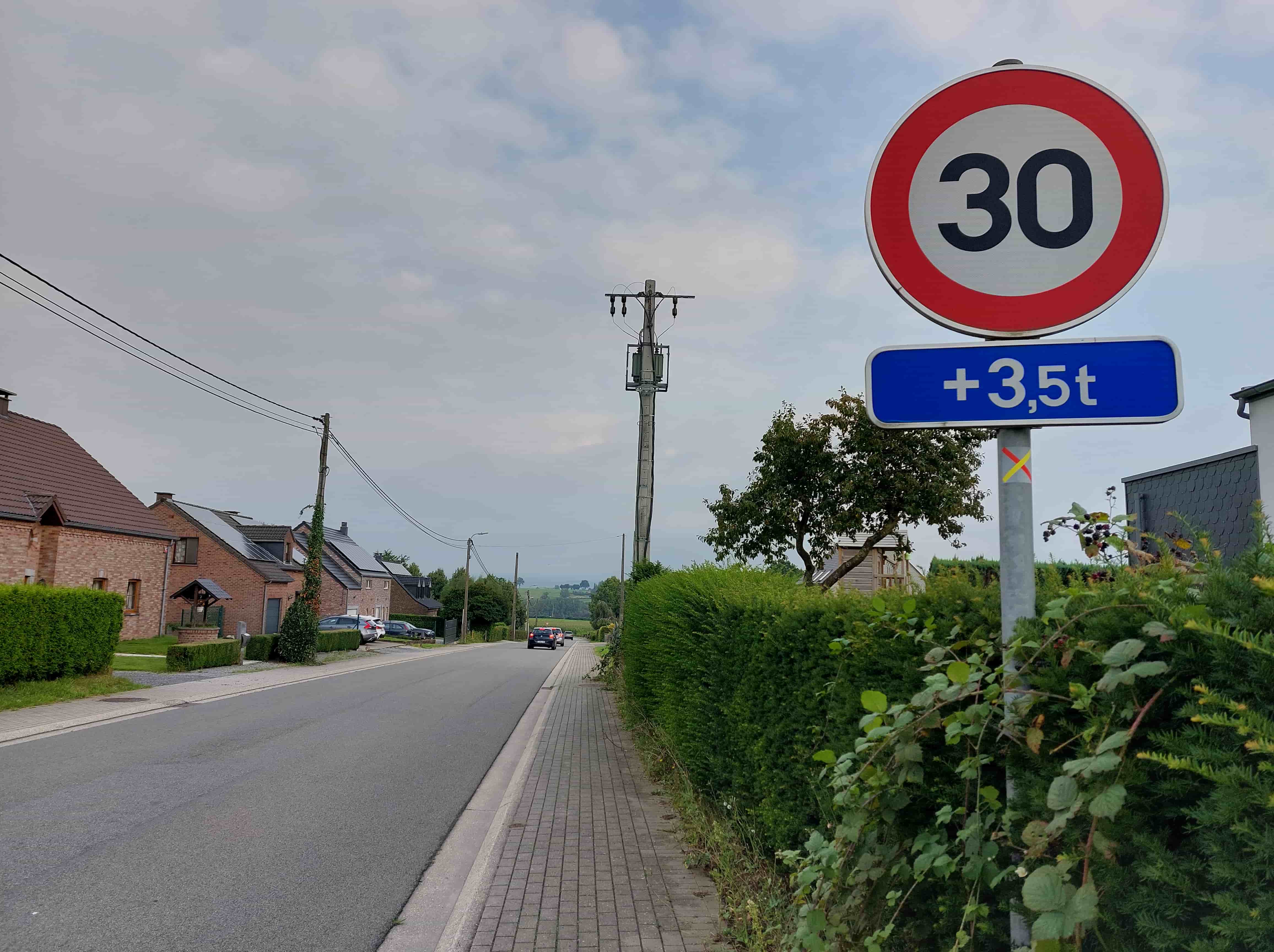 Absolument ! En tant qu'expert en code de la route belge, je vais analyser cette image et le contexte de la question avec précision.
Description détaillée de la situation routière
Type de situation routière:
Nous nous trouvons sur une route asphaltée bidirectionnelle, vraisemblablement à l'intérieur d'une agglomération ou à son entrée, compte tenu des éléments urbains et du libellé de la question. Il s'agit d'une portion de route droite qui s'étend vers l'horizon, bordée de chaque côté par des habitations. Il n'y a pas d'intersection immédiate visible.
Éléments visibles:
Chaussée: La route est en asphalte gris foncé, en bon état. Elle est suffisamment large pour permettre le croisement de deux véhicules, mais ne comporte pas de marquage central, ce qui est courant sur les routes secondaires en agglomération. Une ligne blanche continue délimite le bord droit de la chaussée.
Accotement/Trottoir: Sur le côté droit, un trottoir en pavés gris est clairement délimité par une bordure. Au-delà du trottoir, une haie bien taillée borde la propriété où se trouve le panneau de signalisation.
Bâtiments: Plusieurs maisons d'habitation en briques sont visibles, principalement sur le côté gauche de la route. Elles présentent des toits en tuiles marron, et certaines sont équipées de panneaux solaires, ce qui confirme un environnement résidentiel. Une haie dense et des arbustes se trouvent sur le côté droit, masquant partiellement une habitation.
Véhicules: Deux voitures sont stationnées sur le côté gauche de la chaussée, plus loin dans la perspective. Deux autres véhicules (une voiture sombre et une voiture plus claire derrière elle) circulent sur la chaussée, dans le même sens que le conducteur supposé de la question, et s'éloignent dans la distance.
Poteaux et câbles: Plusieurs poteaux électriques en bois supportant des lignes aériennes traversent le paysage, renforçant l'idée d'une zone habitée. Un poteau plus robuste, probablement en béton, avec des transformateurs, est visible plus loin.
Végétation: Des haies, des arbres et des arbustes (notamment des ronces avec des mûres) sont présents des deux côtés de la route, ajoutant un caractère semi-rural à l'agglomération.
Ciel: Le ciel est couvert de nuages, mais la luminosité est suffisante, indiquant une journée sans problème de visibilité lié aux conditions météorologiques.
Position et direction des véhicules s'il y en a:
Les deux voitures garées sont sur le côté gauche de la route, parallèles au trottoir, et semblent orientées dans le même sens que le flux de circulation de la question.
Les deux véhicules en mouvement sont dans le lointain, sur la chaussée, circulant également dans le sens de la progression de la route, s'éloignant de l'observateur.
Panneaux de signalisation présents et leur signification (Belgique):
Sur le côté droit de la chaussée, fixés sur le même poteau métallique, nous voyons deux panneaux :
Panneau supérieur (rond à bord rouge, fond blanc, "30" en noir): Il s'agit du panneau C43 "Limitation de vitesse" qui indique que la vitesse maximale autorisée est de 30 km/h.
Panonceau inférieur (rectangulaire bleu, "+3,5t" en blanc): Il s'agit d'un panonceau de catégorie M2 qui précise la signification du panneau principal. Le "+3,5t" signifie "véhicules ou groupes de véhicules dont la masse maximale autorisée est supérieure à 3,5 tonnes".
Signification combinée des panneaux (très importante pour la question) : La limitation de vitesse à 30 km/h ne s'applique spécifiquement qu'aux véhicules ou groupes de véhicules dont la masse maximale autorisée (MMA) est supérieure à 3,5 tonnes. Pour tous les autres véhicules, c'est la règle générale de la zone dans laquelle ils se trouvent qui s'applique. Puisque la question mentionne "dans cette agglomération", la vitesse générale autorisée en agglomération en Belgique est de 50 km/h, sauf indication contraire.
Tout autre élément pertinent pour comprendre la situation:
Le poteau de signalisation comporte également un petit autocollant jaune et orange en forme de "X", qui est probablement une marque technique ou de repérage pour les services d'entretien, et non une signalisation routière. La présence de mûres sur les ronces peut indiquer la période de l'année (fin de l'été/début de l'automne).
Analyse du contexte de la question :
Question: "Dans cette agglomération, je circule à 45 km/h au volant d'une camionnette d'une MMA de 3500 kg. Suis-je en infraction ?"
Mon véhicule: Camionnette d'une MMA de 3500 kg (soit 3,5 tonnes).
Vitesse: 45 km/h.
Lieu: Agglomération.
Signalisation spécifique: Panneau "30" avec panonceau "+3,5t".
Conclusion préliminaire pour la réponse à la question (non demandée dans la description, mais utile pour l'expert):
La camionnette a une MMA de 3500 kg (3,5 tonnes). Le panonceau "+3,5t" signifie que la limitation de vitesse à 30 km/h s'applique uniquement aux véhicules dont la MMA est supérieure à 3,5 tonnes. Puisque la MMA de la camionnette est de 3,5 tonnes (et non supérieure à 3,5 tonnes), la limitation à 30 km/h ne la concerne pas.
En agglomération, la vitesse maximale autorisée est de 50 km/h, sauf indication contraire. Dans ce cas, l'indication contraire ne s'applique pas à notre véhicule.
Le conducteur roule à 45 km/h, ce qui est inférieur à 50 km/h. Par conséquent, il n'est pas en infraction.