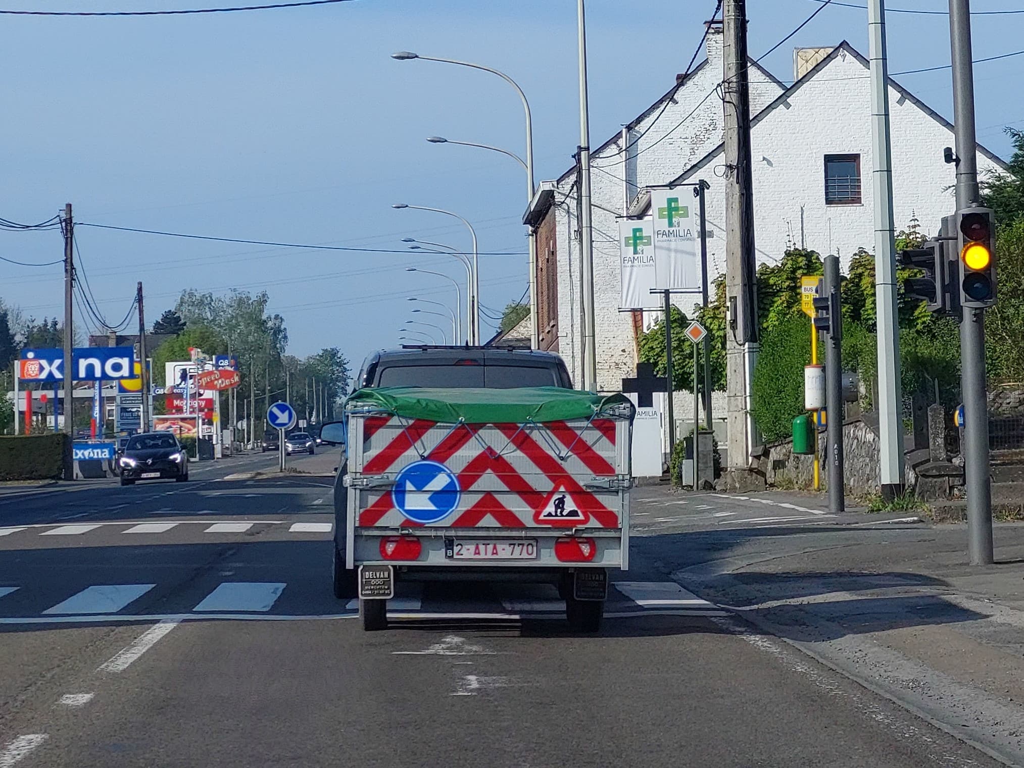 En tant qu'expert en code de la route belge, voici une description détaillée de la situation présentée dans l'image, en relation avec la question posée :
Contexte de la question : "Je circule à 20 km/h, combien de mètres vais-je avoir besoin afin de m'immobiliser devant le feu jaune-orange fixe qui vient de s'allumer ?"
Description détaillée de la situation routière :
Type de situation routière :
La scène se déroule sur une chaussée principale, vraisemblablement en agglomération ou à l'entrée d'une zone urbanisée, caractérisée par la présence de commerces (enseignes Q8, Ixina, Lidl visibles au loin à gauche) et d'habitations. Le véhicule du conducteur (depuis lequel la photo est prise) approche d'une intersection majeure contrôlée par des feux de signalisation lumineux. Immédiatement avant cette intersection, se trouve un passage pour piétons. Une rue latérale ou une entrée semble exister sur la droite, près de laquelle un arrêt de bus est signalé. La route est bordée de trottoirs et de poteaux d'éclairage public.
Éléments visibles :
Chaussée et marquages au sol :
La chaussée est asphaltée et semble sèche.
Un passage pour piétons est clairement visible juste devant le véhicule en face. Il est marqué par des bandes blanches longitudinales (zébra) et deux lignes transversales blanches continues délimitant le début et la fin du passage.
Plusieurs mètres avant le passage pour piétons, on distingue des marques au sol (flèches de direction ou marquages de voie) mais elles ne sont pas entièrement lisibles.
Une ligne blanche continue délimite le bord droit de la chaussée.
Plus loin sur la gauche, des lignes discontinues marquent probablement l'axe de la chaussée ou des voies de circulation.
Véhicules :
Véhicule du conducteur : Implicite, il est le point de vue de la photo.
Véhicule en amont (devant nous) : Un pick-up de couleur sombre tirant une petite remorque à un essieu. La remorque est bâchée en vert et présente à l'arrière une signalisation de grande visibilité avec des chevrons diagonaux rouges et blancs. Elle porte une plaque d'immatriculation belge (2-ATA-770).
Véhicules venant en sens inverse : Une voiture noire avec les phares allumés est visible au loin sur la voie opposée, suivie par d'autres véhicules.
Signalisation lumineuse :
Un feu tricolore vertical est visible sur le côté droit de la chaussée, monté sur un poteau. Il est actuellement allumé en jaune-orange fixe. Un feu piéton est également présent sous le feu tricolore mais son état n'est pas clairement discernable (il semble éteint ou rouge, mais non pertinent pour la question).
Panneaux de signalisation :
Sur la remorque du véhicule devant :
Un panneau circulaire bleu avec une flèche blanche pointant vers la droite (panneau de prescription D1a "Passer à droite de l'obstacle"). Ce panneau, bien que réglementaire, est ici apposé sur un véhicule et peut indiquer des consignes spécifiques pour le dépassement de cet ensemble.
Un panneau triangulaire à fond blanc avec bord rouge et un pictogramme noir représentant un ouvrier en train de creuser (panneau de danger A39 "Travaux").
Sur la droite de la chaussée :
Sous les bannières publicitaires, un petit panneau en forme de losange orange est visible, mais trop petit et partiellement masqué pour être identifié avec certitude comme un panneau réglementaire du code de la route belge.
Un panneau rectangulaire jaune avec le texte "BUS 70 77" indique un arrêt de bus.
Plus loin sur la gauche (en face) : Un panneau circulaire bleu avec une flèche blanche pointant vers le bas et la droite (probablement D1b "Passer à droite de l'obstacle" ou direction obligatoire), indiquant une contrainte de circulation future.
Infrastructures et environnement :
Sur la droite, un bâtiment de couleur blanche avec des bannières verticales affichant "FAMILIA Pharmacie / Centre Orthopédique".
Plusieurs poteaux électriques et lignes aériennes.
Des lampadaires d'éclairage public s'alignent le long de la route.
Des arbres et de la végétation sont présents des deux côtés de la route.
Sur la gauche, au loin, des enseignes commerciales (Ixina, Q8, Lidl) sont visibles, confirmant une zone d'activité.
Un muret en pierres et des buissons le long du trottoir droit.
Position et direction des véhicules :
Le véhicule du conducteur et le pick-up avec remorque le précédant circulent sur la même voie, en ligne droite, en direction du feu tricolore et du passage pour piétons. Les véhicules venant en sens inverse circulent sur leur propre voie, également en ligne droite.
Panneaux de signalisation présents et leur signification (pertinents pour la question) :
Feu jaune-orange fixe : En Belgique, ce feu signifie "arrêt obligatoire", sauf si l'immobilisation du véhicule ne peut se faire dans des conditions de sécurité suffisantes. Étant donné que le feu "vient de s'allumer" et que la vitesse est de 20 km/h, il est attendu du conducteur qu'il s'arrête en toute sécurité.
Passage pour piétons (marquage au sol) : Ce marquage impose au conducteur de laisser la priorité aux piétons qui s'y engagent ou sont sur le point de s'y engager. En cas d'arrêt au feu, le véhicule doit s'immobiliser avant* ce passage pour ne pas l'encombrer et permettre aux piétons de traverser.
Tout autre élément pertinent pour comprendre la situation :
La question insiste sur la vitesse de 20 km/h et le fait que le feu jaune-orange "vient de s'allumer". À cette faible vitesse, un conducteur devrait normalement être en mesure de s'immobiliser en toute sécurité. Le point d'arrêt pertinent pour la question est la ligne d'arrêt qui précède le passage pour piétons, situé juste avant le feu de signalisation. Les panneaux sur la remorque (travaux, flèche) et ceux plus loin sur la route ne sont pas directement pertinents pour la décision immédiate d'arrêter ou non au feu jaune-orange. La route est en bon état et le temps est clair et ensoleillé, suggérant des conditions de conduite optimales.