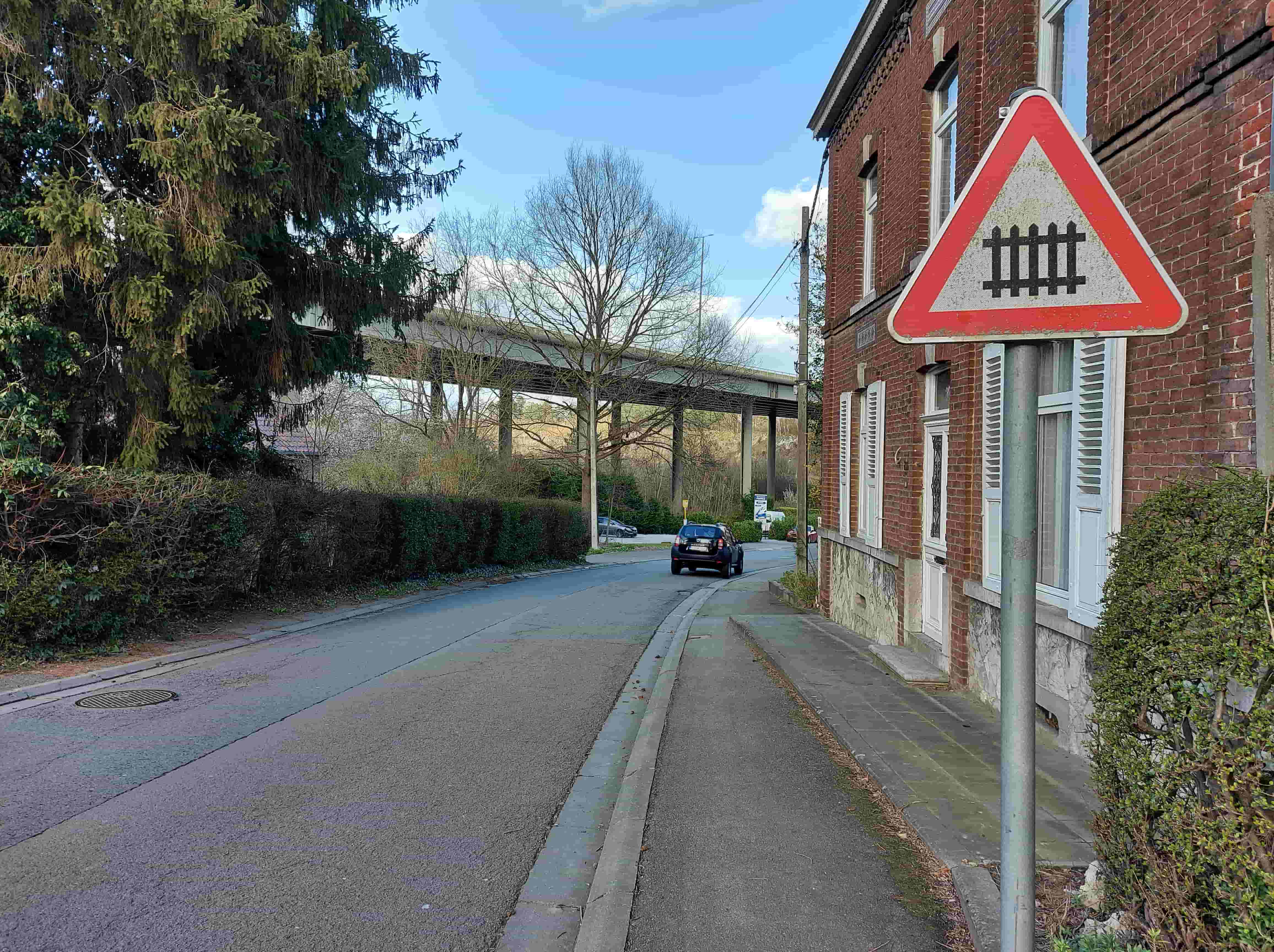 En tant qu'expert en code de la route belge, voici une description détaillée de l'image, analysée dans le contexte de la question d'examen de conduite.
Description Détaillée de la Situation Routière pour la Question d'Examen
Type de situation routière :
L'image présente une portion de voie publique bidirectionnelle (route à double sens) en dehors d'une agglomération dense, ou à sa périphérie. Il s'agit d'une route asphaltée, avec des trottoirs et des bordures de chaque côté. L'environnement est semi-urbain à rural, caractérisé par la présence de bâtiments résidentiels d'un côté et de végétation dense de l'autre, ainsi qu'une infrastructure de grande envergure (viaduc) en arrière-plan. La route s'éloigne du point de vue de l'observateur en décrivant une légère courbe vers la droite.
Les éléments visibles :
La chaussée : Elle est en asphalte, sans marquages au sol centraux visibles (ligne médiane) ou latéraux (bandes de rives), ce qui est courant sur des routes secondaires. Une bouche d'égout est visible sur le côté gauche de la chaussée dans le premier plan.
Les trottoirs : Un trottoir en dalles grises est clairement visible sur le côté droit de la route, devant la maison. Un autre trottoir semble exister sur la gauche, mais il est largement masqué par la végétation.
Les véhicules : Une voiture de couleur foncée (qui ressemble à un SUV Dacia Duster) est visible sur la chaussée, s'éloignant du point de vue de l'observateur. Elle se trouve à une distance moyenne dans la perspective de la photo.
Les panneaux de signalisation : Un unique et très visible panneau de signalisation est présent sur le côté droit de la chaussée, fixé sur un poteau métallique.
Les feux : Aucun feu de signalisation (trilogie ou clignotant) n'est visible sur la route ni en rapport avec un passage à niveau. Un lampadaire d'éclairage public est visible au loin sur le côté gauche de la route.
La végétation : De grands arbres à feuilles persistantes (conifères) et des haies denses bordent le côté gauche de la route. Sur la droite, des buissons sont plantés devant la maison. En arrière-plan, sous le viaduc, on observe des arbres à feuilles caduques, actuellement dénudés (suggérant l'hiver ou le début du printemps).
Les bâtiments : Une maison individuelle en briques rouges avec des volets blancs et des encadrements de fenêtres blancs se dresse sur le côté droit de la route.
L'infrastructure : En arrière-plan, un imposant viaduc ou pont en béton armé traverse la scène perpendiculairement à la route. Ce viaduc est supporté par de larges piliers en béton. Il s'agit d'une structure distincte et n'est pas directement lié au "passage à niveau" de la question, mais il fait partie du décor routier. Un poteau électrique ou téléphonique en bois est également visible juste derrière le panneau de signalisation, avec des câbles suspendus.
Conditions météorologiques : Le ciel est partiellement nuageux avec de larges zones de ciel bleu, indiquant un temps clair et ensoleillé.
Position et direction des véhicules :
Le véhicule sombre est positionné sur la voie de droite (par rapport au sens de circulation en Belgique), roulant dans la même direction que l'observateur (s'éloignant du point de vue). Il est à une certaine distance, en train de négocier un léger virage vers la droite de la route.
Les panneaux de signalisation présents et leur signification :
Panneau A7 : Il s'agit d'un panneau de danger triangulaire à fond blanc, bordé de rouge, avec un pictogramme noir représentant une barrière de chemin de fer stylisée (quatre montants verticaux reliés par une barre horizontale à leur base). Ce panneau, selon le Code de la route belge, est le signal A7 "Passage à niveau avec barrières". Il avertit les conducteurs de la présence imminente d'un passage à niveau équipé de barrières automatiques ou manuelles. L'existence de ce panneau implique la nécessité de redoubler de prudence, de ralentir et d'être prêt à s'arrêter si les barrières sont fermées ou en train de se fermer.
Tout autre élément pertinent pour comprendre la situation :
Distance du danger (contexte de la question) : La question demande "À quelle distance se situe ce danger ?". Selon l'Article 9.1.2 du Code de la route belge, les signaux de danger sont placés à une distance déterminée du danger qu'ils signalent :
En agglomération : 150 mètres avant le danger.
Hors agglomération : 200 mètres avant le danger.
Si la distance est différente, elle doit être précisée par une balise de distance (J1, J2, J3) placée sous le panneau, ou par un panneau additionnel indiquant la distance. Aucun de ces panneaux additionnels n'est visible sous le panneau A7.
• Inférence sur l'agglomération : L'environnement (présence de végétation dense, route sans marquage spécifique, absence de rangées continues de bâtiments urbains, présence d'un viaduc en arrière-plan qui suggère une infrastructure majeure souvent en périphérie) tend à indiquer que l'on se trouve hors agglomération ou à sa limite. Par conséquent, la distance de 200 mètres est la plus probable en l'absence d'indication contraire.
En résumé, l'image montre un conducteur averti par un panneau A7 (passage à niveau avec barrières) d'un danger situé, en l'absence de balises de distance complémentaires, à la distance réglementaire de 200 mètres, compte tenu de l'environnement qui semble être hors agglomération.