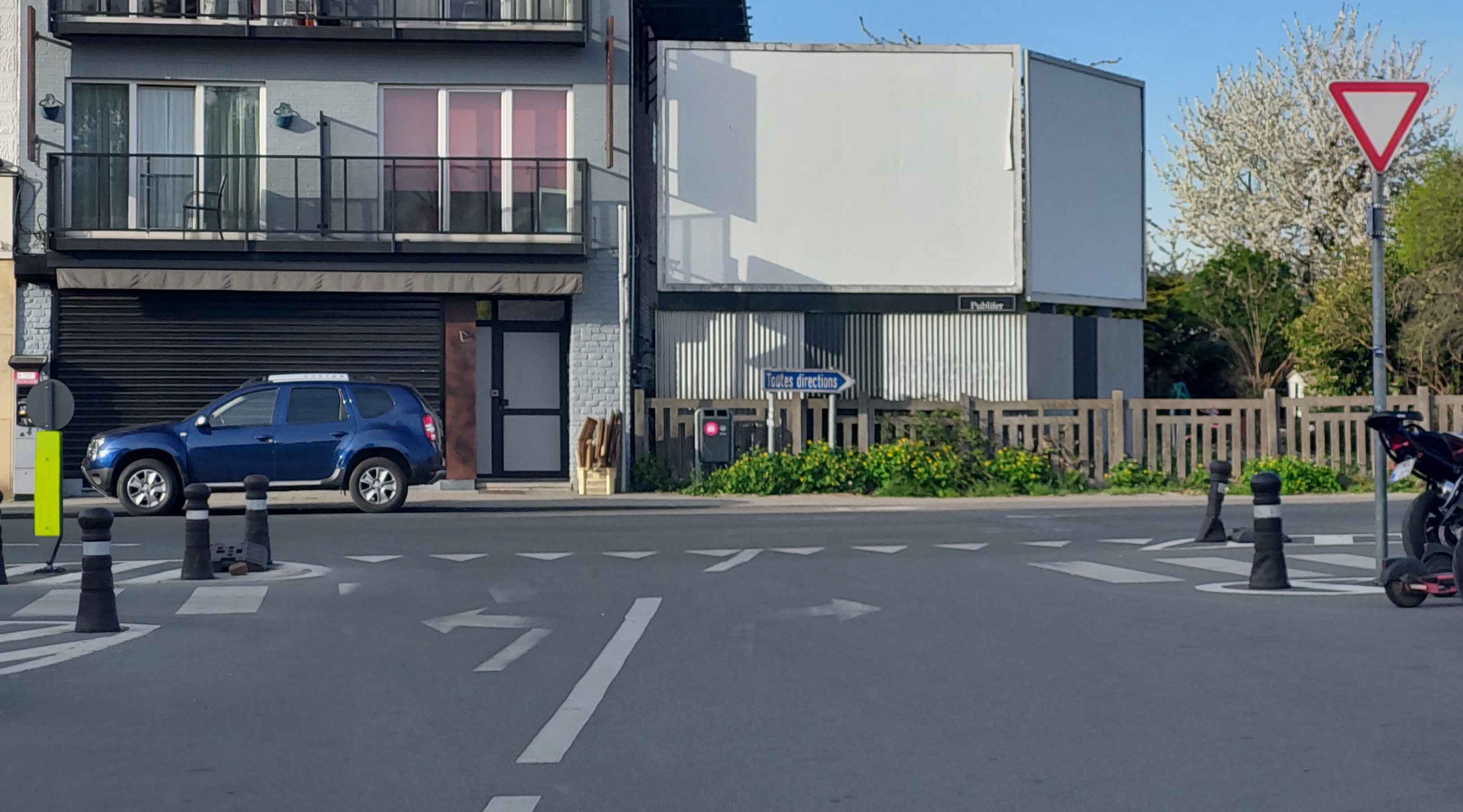 En tant qu'expert en code de la route belge, voici une analyse détaillée de l'image, en tenant compte du contexte fourni ("J'ai été quelque peu distrait et je veux tout de suite virer à gauche au prochain carrefour.") :
Description détaillée de la situation routière:
Type de situation routière:
L'image présente une intersection routière (un carrefour) en milieu urbain ou semi-urbain, vue depuis la perspective du conducteur qui approche de ce carrefour. La route sur laquelle se trouve le conducteur semble être une voie bidirectionnelle avec des aménagements pour les piétons (trottoirs, passages piétons). La signalisation indique clairement que le conducteur se trouve sur une route où il doit céder le passage à l'intersection.
Éléments visibles:
Véhicules:
Un SUV Dacia Duster bleu est stationné sur le côté gauche de la chaussée, avant l'intersection, face à la direction de la marche (donc éloigné de la caméra).
Une partie d'une moto ou d'un scooter (avec plaque d'immatriculation) est visible sur le trottoir à l'extrême droite.
Une trottinette électrique (couleur rose/noire) est également stationnée sur le trottoir à l'extrême droite.
Il n'y a pas d'autres véhicules en mouvement visibles dans l'image.
Panneaux de signalisation:
Un panneau B1 "Cédez le passage" (triangle inversé à bord rouge et fond blanc) est clairement visible sur un poteau sur le côté droit de la chaussée, avant l'intersection.
Un panneau indicateur de direction bleu, avec une flèche pointant vers la droite et l'inscription "Toutes directions", est situé plus loin sur la droite, au niveau de l'intersection.
Feux de signalisation:
Aucun feu de signalisation (rouge, orange, vert) n'est visible à cette intersection.
Marquages au sol:
Une ligne de "dents de requin" (triangles blancs peints sur la chaussée) est présente juste avant l'intersection, confirmant l'obligation de céder le passage (en accord avec le panneau B1).
Des flèches de sélection de voie sont peintes sur la chaussée dans la voie actuelle du conducteur : une flèche indiquant la direction tout droit et une flèche indiquant la direction à gauche. Ceci signifie que le conducteur peut soit continuer tout droit, soit tourner à gauche depuis cette voie.
Un passage pour piétons (bandes rectangulaires blanches) traverse la chaussée juste avant la ligne de cédez le passage.
Des lignes discontinues délimitent la voie et les aménagements centraux.
Autres éléments:
Bâtiments: Un immeuble de plusieurs étages avec balcons et volets roulants est visible sur la gauche. Plus loin sur la droite, des structures avec de grands panneaux publicitaires blancs.
Végétation: Des arbres (dont un en fleurs blanches, suggérant le printemps) et des parterres de fleurs jaunes bordent la route sur la droite.
Mobilier urbain: Des potelets noirs et blancs délimitent les trottoirs et les aménagements centraux. Une petite borne/poteau vert fluo avec un élément noir est visible sur la gauche. Une borne de couleur rouge foncé (possiblement une borne d'appel d'urgence ou un boîtier électrique) est visible sur un poteau près du panneau "Toutes directions". Une clôture en bois sépare la chaussée de la zone verte sur la droite.
Position et direction des véhicules s'il y en a:
Le SUV bleu est stationné sur la voie de gauche, parallèle au trottoir, face à l'intersection.
La moto/scooter et la trottinette électrique sont stationnées sur le trottoir droit, en dehors de la chaussée.
Le véhicule du conducteur (implicite dans la question) est en approche de l'intersection, dans la voie qui permet de continuer tout droit ou de tourner à gauche.
Panneaux de signalisation présents et leur signification (Code de la route belge):
Panneau B1 "Cédez le passage": Ce panneau triangulaire inversé (rouge et blanc) indique que le conducteur doit céder le passage à tous les conducteurs qui circulent sur la voie ou les voies transversales, quelle que soit leur direction. L'arrêt n'est pas obligatoire si le passage est libre, mais le conducteur doit réduire sa vitesse et, si nécessaire, s'arrêter pour laisser passer les usagers prioritaires.
Panneau "Toutes directions": Ce panneau indicateur (bleu avec flèche blanche) est un panneau de localisation ou de direction. Il indique que la direction pointée par la flèche (ici, vers la droite, probablement la route transversale) donne accès à "toutes directions", signifiant souvent qu'il s'agit d'une artère importante ou d'une route permettant de rejoindre de nombreuses destinations.
Tout autre élément pertinent pour comprendre la situation:
La présence des flèches de sélection de voie (tout droit et gauche) est capitale, car la question indique que le conducteur veut "virer à gauche". Ces flèches confirment que le virage à gauche est une option depuis la voie actuelle du conducteur.
Le passage pour piétons juste avant la ligne de cédez le passage signifie que le conducteur devra être particulièrement attentif aux piétons qui pourraient traverser, et leur céder le passage avant même de se préoccuper du trafic traversant.
L'îlot central (matérialisé par les potelets et les marquages) au milieu de l'intersection influence la trajectoire et le rayon de braquage lors d'un virage.
Le fait que le conducteur ait été "quelque peu distrait" est un élément psychologique crucial. Dans une situation où il faut céder le passage et gérer un virage à gauche (qui est souvent plus complexe qu'un virage à droite en termes de priorités et de véhicules venant en sens inverse), la distraction augmente considérablement le risque d'erreur.
En résumé, la situation montre un conducteur s'approchant d'un carrefour où il doit céder le passage, avec la possibilité de tourner à gauche, et où un passage pour piétons est présent juste avant l'intersection. La signalisation au sol confirme les options de direction, tandis que le panneau B1 dicte la règle de priorité.