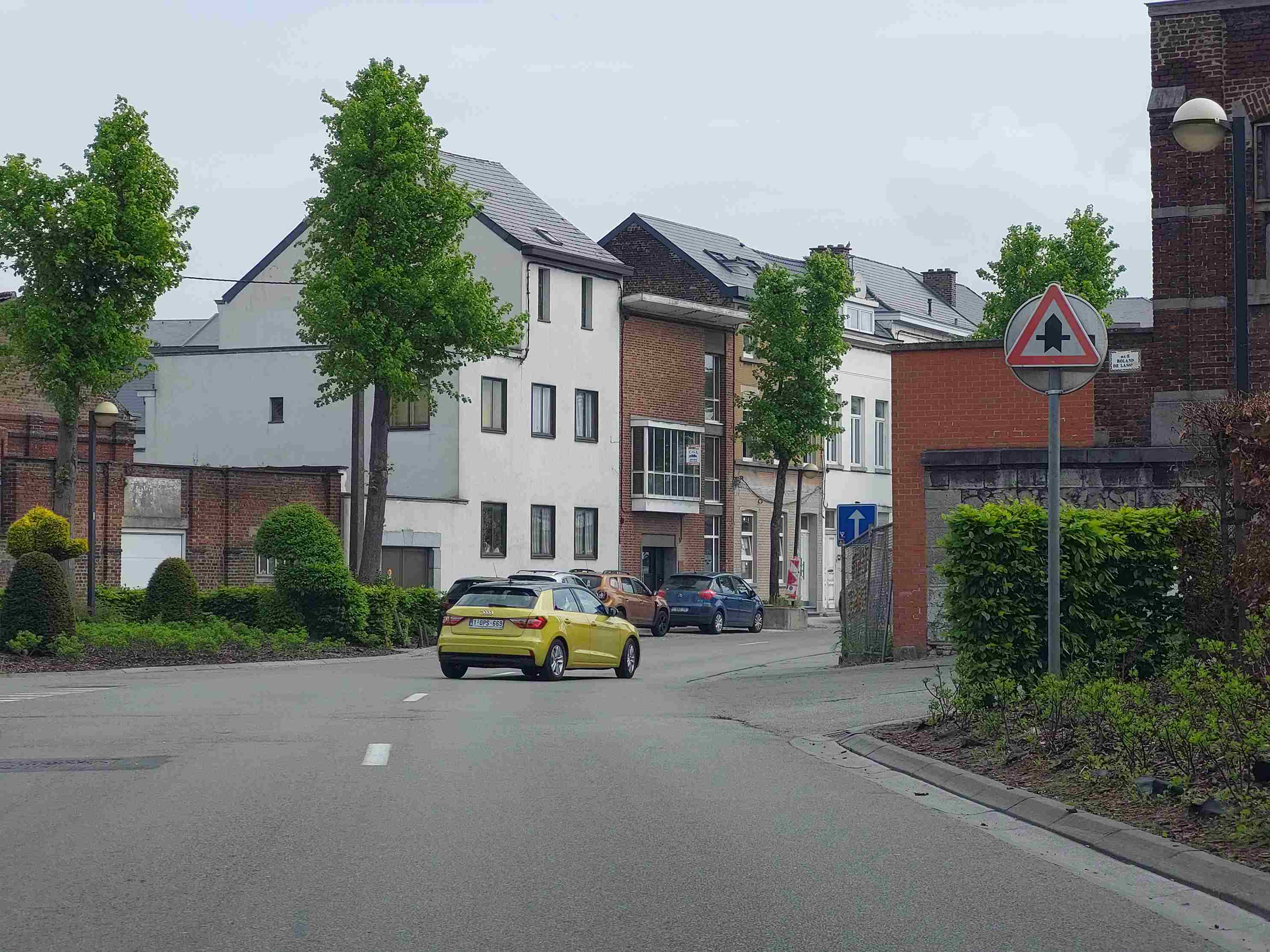 Voici une description détaillée de l'image, en tant qu'expert en code de la route belge :
Description détaillée de la situation routière :
Type de situation routière :
L'image présente une vue de la perspective d'un conducteur circulant sur une route en milieu urbain ou semi-urbain. Le conducteur est en approche d'un carrefour ou d'une intersection, clairement visible plus loin sur la gauche et potentiellement aussi à droite (bien que moins distinct). La rue est bordée de maisons, d'arbres et de végétation, suggérant une zone habitée.
Éléments visibles :
Chaussée : La route est asphaltée, large d'au moins deux voies (une dans chaque direction, ou une voie large permettant le dépassement/l'évitement des véhicules stationnés). Une ligne discontinue blanche est visible sur la chaussée, marquant le centre théorique de la voie de circulation du conducteur ou délimitant une bande de stationnement inexistante sur ce côté.
Marquages au sol : Outre la ligne discontinue blanche, aucun autre marquage au sol de priorité (comme un triangle blanc "céder le passage" ou une ligne d'arrêt) n'est visible directement sur la chaussée du conducteur en approche du carrefour.
Panneaux de signalisation :
Principal : Sur le côté droit de la chaussée, juste avant le carrefour, se trouve un panneau de signalisation triangulaire à bord rouge et fond blanc. Au centre de ce panneau figure une large flèche verticale noire (représentant la route principale sur laquelle le conducteur circule) traversée par une fine flèche horizontale noire avec une petite barre noire à son extrémité droite (représentant une route secondaire venant de la droite).
Secondaire : Plus loin dans l'intersection ou sur la rue latérale gauche, on aperçoit un panneau carré bleu avec une flèche blanche pointant vers le haut. Il semble s'agir d'un panneau d'obligation de direction.
Pancarte de rue : Derrière le panneau principal, sur le mur en briques, une pancarte indique "RUE ROLAND DE LASSO".
Véhicules :
Une voiture jaune/vert citron (ressemblant à une Audi A1) est visible au centre de l'image, s'éloignant du point de vue de la caméra, circulant sur la même voie. Sa plaque d'immatriculation est 1-GPS-669.
Plusieurs véhicules sont stationnés sur le côté gauche de la rue, plus loin vers l'intersection (par exemple, une voiture bleu foncé, une voiture brun-orangé).
Bâtiments : Des maisons de style varié bordent la route des deux côtés, avec des façades en briques ou crépies en blanc.
Végétation : Des arbres matures et des buissons d'ornement sont présents le long de la route et des propriétés.
Éclairage public : Un lampadaire de type "boule" est visible sur le côté droit, juste derrière le panneau principal.
Mobilier urbain : Trottoirs, bordures de trottoir, avaloir/grille d'évacuation des eaux.
Position et direction des véhicules :
Le véhicule du conducteur (impliqué par la perspective de l'image) est sur la voie de droite, avançant tout droit vers le carrefour.
La voiture jaune circule devant le véhicule du conducteur, également sur la voie de droite et se dirigeant tout droit vers l'intersection.
Les autres véhicules sont stationnés le long de la rue, à gauche, et n'impactent pas directement la situation de priorité au carrefour.
Panneaux de signalisation présents et leur signification (belge) :
Le panneau principal sur la droite est le panneau de signalisation B9 "Priorité" (Catégorie B - Signaux de priorité).
Signification du B9 : Ce panneau de danger triangulaire (bord rouge, fond blanc, avec les flèches noires) signifie que le conducteur circule sur une route prioritaire. Au prochain carrefour (ou croisement), les usagers venant des routes adjacentes (représentées par les fines flèches horizontales) devront céder le passage. Le conducteur sur cette route a donc la priorité sur les usagers venant des voies transversales.
• Le panneau carré bleu avec la flèche blanche est un panneau F19 ou F17 "Direction obligatoire à suivre" (Catégorie F - Signaux d'indication). Sa présence plus loin dans la rue latérale indique une direction imposée pour les usagers de cette rue, mais n'a pas d'impact direct sur la priorité au carrefour pour le conducteur venant de la route principale.
Tout autre élément pertinent :
Le contexte de la question d'examen ("Ce signal signifie qu'au prochain carrefour, je suis prioritaire sur les conducteurs qui viennent :") met en évidence l'importance du panneau B9. Il est clair que le conducteur doit interpréter ce panneau pour déterminer sa situation de priorité au carrefour imminent. La visibilité générale est bonne, les conditions météorologiques semblent être couvertes mais sans précipitations.