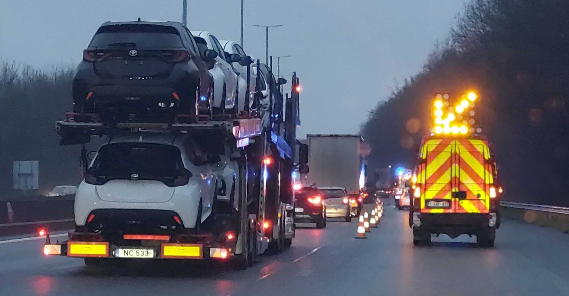 En tant qu'expert du code de la route belge, voici une description détaillée de l'image pour comprendre le contexte de la question d'examen de conduite :
Description détaillée de la situation routière :
Type de situation routière :
La scène se déroule sur une autoroute ou une voie rapide à plusieurs bandes de circulation, dans des conditions de faible luminosité (crépuscule ou nuit) et sur une chaussée humide, potentiellement après la pluie. Il s'agit d'une situation de congestion ou de ralentissement du trafic due à une intervention ou des travaux sur la bande de droite, entraînant la fermeture de cette bande et la nécessité pour les conducteurs de se rabattre.
Éléments visibles :
Véhicules :
Au premier plan et sur la bande de gauche : Un imposant camion porte-voitures, chargé de plusieurs véhicules neufs (des citadines, principalement des Toyota Yaris Cross, une noire en haut et une blanche en bas visible de l'arrière). Les feux arrière, les feux de gabarit et les dispositifs réfléchissants du camion sont allumés et clairement visibles. La plaque d'immatriculation visible est "NC 533-".
Plus en avant et sur la bande de droite : Un véhicule de service de couleur jaune et rouge à bandes réfléchissantes (type fourgonnette ou petit utilitaire de signalisation de chantier/intervention) est positionné. Ses feux de détresse et ses gyrophares sont probablement actifs, mais surtout, un panneau lumineux en forme de flèche oblique pointant vers la gauche est monté sur son toit. La plaque d'immatriculation visible est "1-BZY-811".
Autres véhicules : De nombreux autres véhicules sont visibles devant et autour du camion porte-voitures, principalement sur les bandes du milieu et de droite, avec leurs feux arrière allumés, suggérant un trafic dense et potentiellement ralenti. Un semi-remorque blanc est visible plus loin sur la bande de gauche.
Marquages au sol et signalisation temporaire :
Bandes de circulation : Au moins deux à trois bandes de circulation sont visibles dans le sens de la marche. Le camion porte-voitures se trouve sur la bande la plus à gauche des bandes actives. Le véhicule de service est positionné sur la bande de droite.
Balises (cônes) : Une ligne de cônes de signalisation orange et blancs est visible sur la bande de droite, s'étendant depuis l'arrière du véhicule de service et marquant la fermeture progressive de cette bande.
Chaussée : La chaussée est visiblement mouillée, créant des reflets des feux des véhicules et de l'éclairage.
Éléments environnementaux :
Des arbres ou de la végétation bordent la route sur la droite.
Des poteaux d'éclairage ou des portiques sont visibles au-dessus des bandes de circulation.
Une glissière de sécurité est visible sur l'accotement droit.
Position et direction des véhicules :
Tous les véhicules visibles circulent dans la même direction. Le photographe se trouve derrière le camion porte-voitures, dans la même bande de circulation (celle de gauche). Le camion porte-voitures avance également dans cette bande. Le véhicule de service est immobilisé ou roule très lentement sur la bande de droite, servant de véhicule d'annonce de la fermeture de cette bande. Les autres véhicules en arrière-plan avancent également dans la même direction, s'adaptant à la situation de ralentissement et de rabattement.
Panneaux de signalisation présents et leur signification :
Le panneau de signalisation le plus pertinent et visible est le panneau lumineux en forme de flèche oblique pointant vers la gauche monté sur le toit du véhicule de service. Ce type de panneau (souvent associé aux panneaux J25 ou J26 en Belgique pour les signaux d'indication de danger temporaire) a pour signification d'indiquer clairement aux conducteurs de la bande de circulation où il est placé (ici, la bande de droite) qu'ils doivent changer de bande en se rabattant vers la gauche. Il s'agit d'une instruction claire de déviation de la circulation.
Tout autre élément pertinent pour comprendre la situation :
La question d'examen indique : "Je suis obligé de changer de bande de circulation, les conducteurs de la bande adjacente (celle du milieu) :". En combinant cette information avec l'image, on comprend que :
Le conducteur (vous) se trouve dans la bande de droite (celle qui est signalée comme fermée par le véhicule de service et les cônes).
Vous êtes donc obligé de vous insérer dans la bande de circulation immédiatement à votre gauche, qui est désignée comme "celle du milieu" dans la question.
• La situation illustre donc un cas de rabattement forcé dû à une bande de circulation fermée, et la question porte sur les obligations ou comportements attendus des conducteurs dans la bande adjacente (celle du milieu) où vous devez vous insérer.
