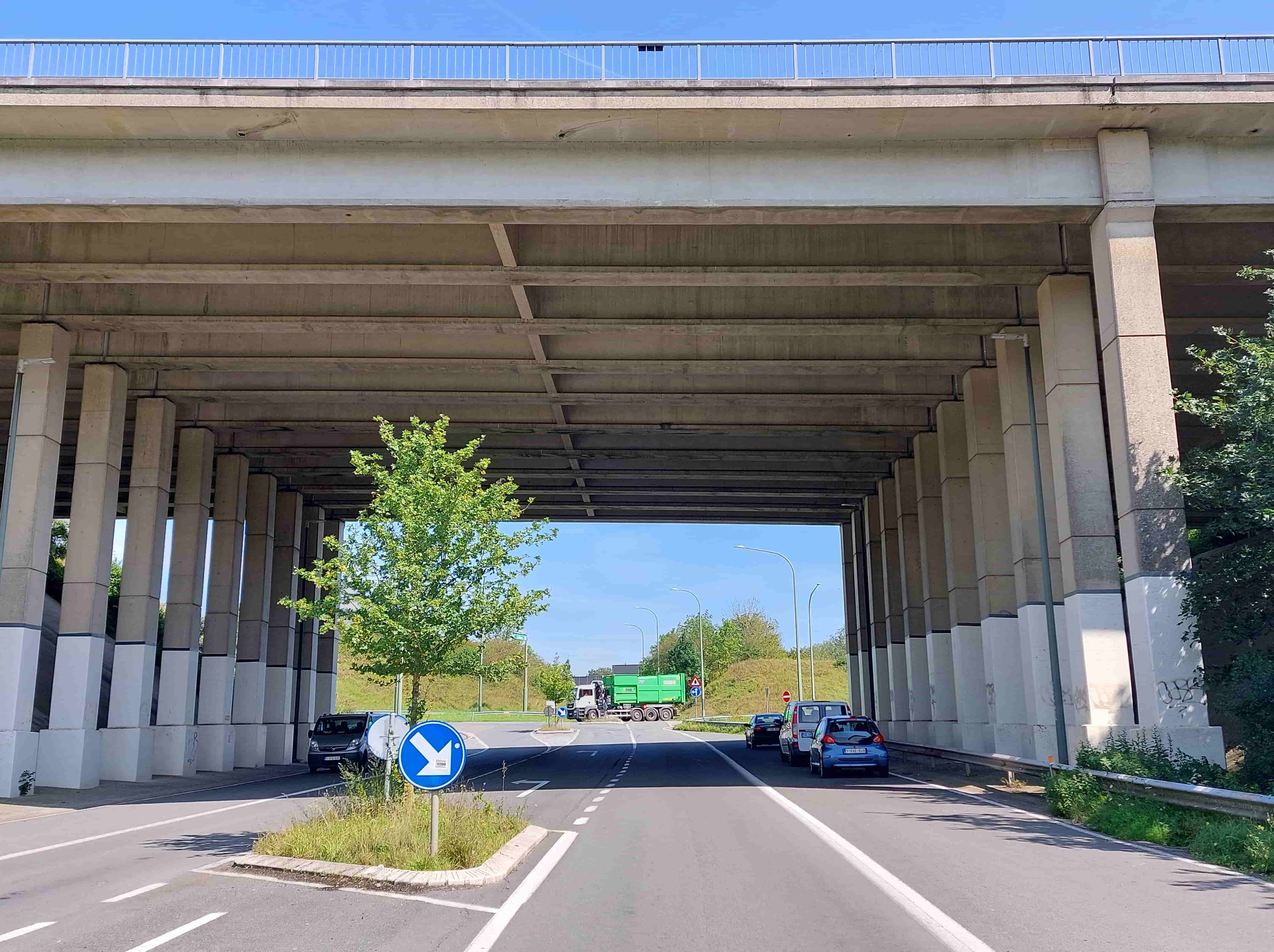 Absolument ! En tant qu'expert en code de la route belge, je vais analyser cette image pour vous fournir une description détaillée, essentielle à la compréhension du contexte de la question d'examen.
Description détaillée de la situation routière
Nous sommes face à une section de route qui passe sous un imposant viaduc ou pont d'autoroute en béton. L'angle de prise de vue est celui du conducteur d'un véhicule qui s'apprête à traverser cette structure et à aborder une intersection complexe juste après.
Type de situation routière:
Il s'agit d'une route bidirectionnelle (bien que le sens de la circulation ne soit pas explicitement indiqué pour les voies au-delà du pont, le marquage au sol et les panneaux lointains suggèrent une complexité) traversant un passage inférieur sous une infrastructure autoroutière majeure. Immédiatement après le passage sous le pont, la route se transforme en une intersection importante, visiblement un carrefour giratoire (rond-point) ou une série de bifurcations avec un îlot central conséquent. La présence de plusieurs véhicules stationnés sur le côté droit suggère une zone potentiellement utilisée pour le stationnement ou l'arrêt.
Les éléments visibles:
Infrastructure générale:
Un large viaduc/pont d'autoroute massif en béton, supporté par de nombreuses rangées de piliers en béton de chaque côté de la chaussée. L'ossature sous le tablier est bien visible.
Un garde-corps métallique blanc est visible au-dessus du pont, délimitant la chaussée supérieure (probablement une autoroute).
La chaussée est en asphalte, en bon état.
Un accotement de terre ou végétalisé est présent sur le côté droit de la route sous le pont, partiellement délimité par une bande de gazon et un rail de sécurité.
Un rail de sécurité métallique est installé le long de l'accotement droit, sous le pont.
Un îlot central végétalisé avec un jeune arbre se trouve sur la gauche de notre voie, juste après la sortie du pont, marquant le début de l'intersection.
Plusieurs lampadaires sont visibles au-delà de l'intersection, suggérant un éclairage public.
Des graffitis sont visibles sur certains piliers en béton sous le pont.
Marquages au sol:
Une ligne blanche continue délimite le bord droit de la chaussée principale, mais elle est précédée d'une ligne blanche discontinue qui sépare la voie de circulation des véhicules stationnés à droite, créant une sorte de bande de stationnement ou d'arrêt.
Une double ligne blanche continue sépare clairement notre voie de la voie adjacente à l'îlot central sur la gauche, indiquant une interdiction de franchissement.
Plusieurs flèches directionnelles au sol sont visibles au loin, au-delà de l'intersection, indiquant des directions de virage (par exemple, une flèche incurvée vers la gauche).
Véhicules:
Sur l'accotement droit, sous le pont: Trois véhicules sont stationnés, tous orientés dans le même sens que notre prise de vue (dos à l'observateur):
Une petite voiture citadine bleue (type Citroën C1 ou Peugeot 107/108), immatriculée "1-KAE-640" (plaque belge).
Une camionnette blanche (type utilitaire), juste derrière la citadine bleue.
Une berline foncée (type BMW série 3), derrière la camionnette blanche.
Plus loin, dans l'intersection: Un gros camion vert avec une cabine blanche est en mouvement, visiblement en train de naviguer dans l'intersection, venant de la gauche de la photo et se dirigeant vers la droite.
Panneaux de signalisation (Code de la route belge):
Sur l'îlot central, à gauche: Un panneau D1 (rond bleu avec flèche blanche oblique vers le bas-droite). Ce panneau signifie "Direction obligatoire à la prochaine intersection" et oblige les conducteurs à passer à droite de l'îlot ou de l'obstacle.
Plus loin, à droite dans l'intersection (proche du camion):
Un panneau D1 (rond bleu avec flèche blanche pointant vers la droite). Il signifie "Direction obligatoire à la prochaine intersection" et impose de tourner à droite.
Un panneau C1 (rond rouge avec une barre blanche horizontale) derrière le D1. Il s'agit du panneau "Sens interdit", interdisant l'accès à tout véhicule dans le sens de la circulation qu'il indique.
Plus loin, à gauche dans l'intersection (proche du camion):
Un panneau A9 (triangle rouge avec un point d'exclamation noir). C'est le panneau "Danger général", signalant un danger non couvert par un panneau spécifique. Il est souvent accompagné d'un panneau additionnel précisant la nature du danger, mais ici, il est illisible.
Position et direction des véhicules:
Les trois voitures garées sur la droite sont stationnées parallèlement à la chaussée, sur ce qui semble être un accotement ou une bande de stationnement dédiée, et sont toutes orientées dans le sens de la marche de l'observateur.
Le camion vert est en train de traverser l'intersection située après le pont, se déplaçant de la gauche vers la droite de l'image.
Panneaux de signalisation et leur signification (Belgique):
Panneau D1 (flèche oblique bas-droite): Obligation de passer à droite de l'obstacle (l'îlot central).
Panneau D1 (flèche droite): Obligation de tourner à droite à l'intersection.
Panneau C1 (Sens interdit): Interdiction formelle d'entrer dans la voie ou la zone signalée dans le sens de la circulation.
Panneau A9 (Danger général): Avertit d'un danger non spécifié, nécessitant une vigilance accrue.
Tout autre élément pertinent pour comprendre la situation:
La question porte sur le stationnement sur "cet accotement à droite sous le pont de l'autoroute". Le fait que plusieurs véhicules y soient déjà stationnés est une observation factuelle, mais n'implique pas nécessairement la légalité du stationnement. En droit belge, le stationnement sous un pont ou dans un passage souterrain est généralement interdit (Article 24.1.3° du code de la route), sauf signalisation contraire (ex: panneau E9a "P" indiquant un parking). L'endroit est bien sous le pont, sur ce qui ressemble à un accotement aménagé ou une voie de service. Le rail de sécurité sur le côté droit renforce l'idée d'un bord de chaussée. L'approche d'une intersection majeure après le pont implique également des règles spécifiques de stationnement à proximité des carrefours.
Cette description complète doit vous permettre de bien appréhender l'environnement et les règles potentielles applicables à la question de stationnement.