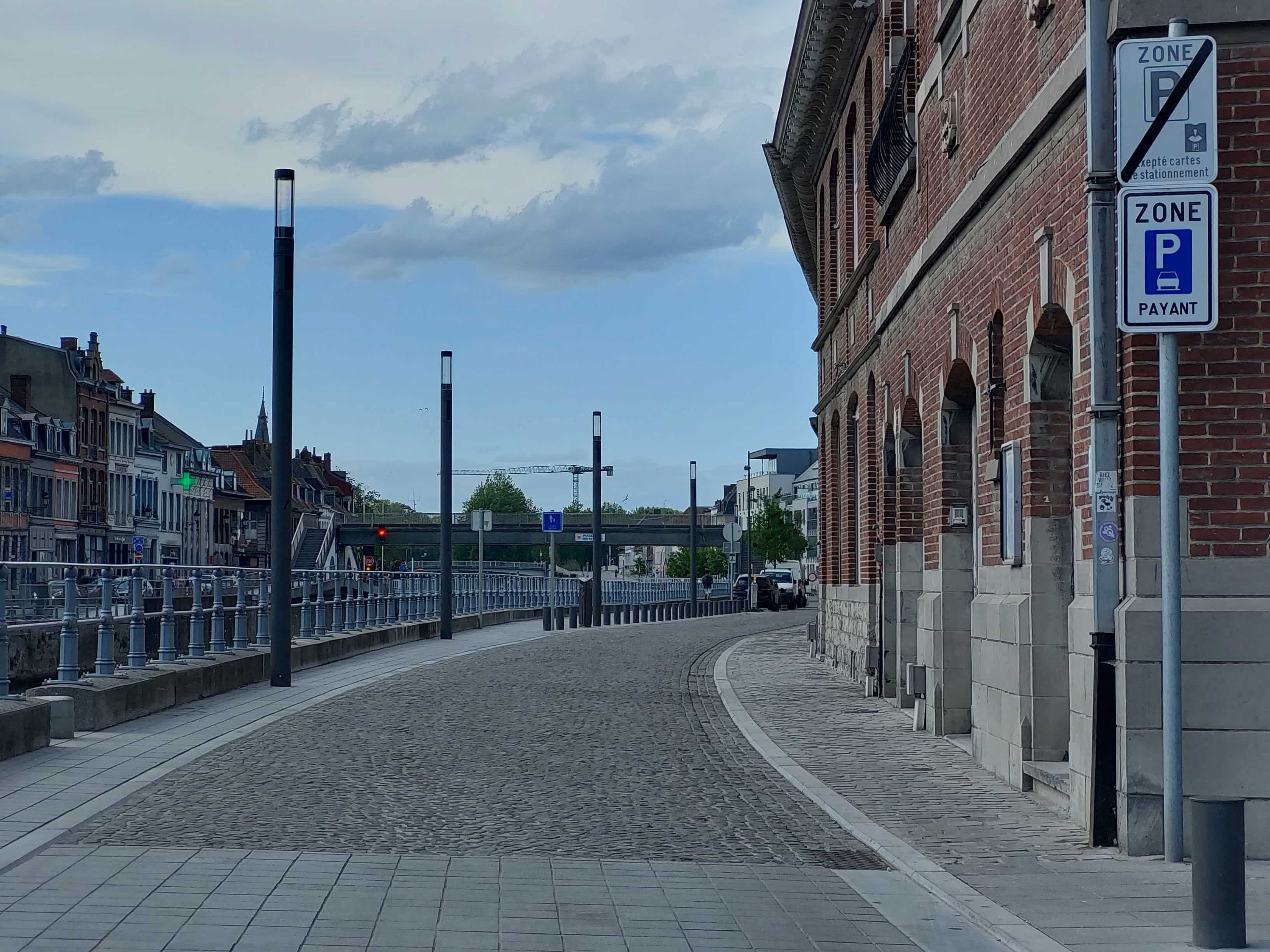 En tant qu'expert en code de la route belge, voici une analyse détaillée de l'image pour comprendre le contexte de la question :
Description détaillée de la situation routière
Type de situation routière:
L'image présente une rue urbaine qui longe ce qui semble être un cours d'eau (canal ou rivière) sur la gauche. Il s'agit d'une zone bâtie dense avec des immeubles anciens typiques d'une ville belge. La route elle-même est en pavés, bordée par un trottoir large à droite le long des bâtiments et un passage piétonnier à gauche, protégé par une rambarde métallique. Plus loin, un pont enjambe le cours d'eau, indiquant une potentielle intersection ou un croisement de voies. La situation est caractéristique d'un environnement urbain où la réglementation du stationnement est souvent spécifique.
Éléments visibles:
Chaussée et Trottoirs: La chaussée principale est revêtue de pavés et s'incurve légèrement vers la droite. Sur la droite, un large trottoir en dalles grises longe un bâtiment. Une fine bordure blanche sépare le trottoir de la chaussée pavée. Sur la gauche, un autre trottoir carrelé, séparé de la chaussée par une rambarde métallique, longe le cours d'eau.
Bâtiments: Sur la droite, un grand bâtiment en briques rouges avec des arches et des éléments architecturaux classiques occupe le premier plan. Sur la gauche, de l'autre côté du canal, on aperçoit une rangée de maisons urbaines colorées, avec une croix verte lumineuse (enseigne de pharmacie) visible au loin.
Panneaux de signalisation: Deux panneaux de zone de stationnement sont empilés sur un poteau gris clair, fixé au bâtiment en briques sur la droite.
Panneau supérieur (E9b - Fin de zone de stationnement): Il s'agit d'un panneau carré blanc bordé de bleu avec un "P" bleu barré d'une ligne diagonale rouge. En dessous du "P", on lit "XEPTÉ CARTES E STATIONNEMENT" (probablement "Excepté cartes de stationnement" ou "Excepté détenteurs de cartes de stationnement"), ce qui indique la fin d'une zone de stationnement réglementée qui autorisait le stationnement sauf pour les détenteurs de cartes spécifiques.
Panneau inférieur (E9a - Début de zone de stationnement réglementé): Il s'agit d'un panneau carré blanc bordé de bleu avec un "P" bleu et un pictogramme de voiture en dessous, accompagné de la mention "PAYANT". Ce panneau indique le début d'une nouvelle zone où le stationnement est réglementé et payant.
Plus loin sur la gauche, avant le pont, on distingue un panneau STOP (B5) et, sous le pont, un panneau carré bleu avec un pictogramme de piéton (probablement F99b - Chemin réservé aux piétons).
Feux de signalisation: Un feu rouge est visible sur la gauche, au niveau du pont, en conjonction avec le panneau STOP.
Véhicules: Plusieurs véhicules (une camionnette blanche, une voiture foncée) sont stationnés ou à l'arrêt plus loin sur la droite de la chaussée, dans la direction de la marche.
Aménagements: Plusieurs réverbères modernes sont alignés le long de la chaussée. Une rambarde métallique longe le côté gauche de la chaussée. Une grue de chantier est visible à l'horizon derrière le pont.
Ciel: Le ciel est nuageux avec des éclaircies.
Position et direction des véhicules s'il y en a:
Les véhicules visibles (camionnette, voiture) sont stationnés parallèlement au trottoir sur la droite, plus loin sur la route, après les panneaux de signalisation principaux. Ils sont orientés dans le sens de la progression de l'observateur (vers le pont). Aucun véhicule n'est visible en mouvement au premier plan.
Panneaux de signalisation présents et leur signification (détaillé):
Panneau supérieur (E9b) : Fin de zone de stationnement réglementé ("ZONE P" barré + "Excepté cartes de stationnement"). Ce panneau signale la fin d'une zone où des règles de stationnement spécifiques (ici, avec des exemptions pour certaines cartes) étaient en vigueur. Il est important de noter qu'il est placé au-dessus du panneau E9a, ce qui peut indiquer que la zone précédente se termine à cet endroit.
Panneau inférieur (E9a) : Début de zone de stationnement réglementé ("ZONE P" + pictogramme voiture + "PAYANT"). Ce panneau indique que, à partir de l'endroit où il est implanté, la rue ou la zone concernée est soumise à une réglementation de stationnement. Dans ce cas précis, la mention "PAYANT" signifie que tout stationnement autorisé dans cette zone est sujet à paiement (généralement via un horodateur ou une application mobile).
Interprétation combinée des panneaux de zone: La configuration des deux panneaux empilés est cruciale. Le panneau inférieur (E9a "PAYANT") annule et remplace la règle précédente (E9b barré) et s'applique pour la portion de route au-delà de ces signaux. On entre donc dans une nouvelle zone de stationnement où le paiement est requis.
Panneau B5 : STOP. Oblige les conducteurs venant de la voie où ce panneau est implanté à marquer un arrêt complet à la ligne d'arrêt ou, à défaut, à l'aplomb du signal, et à céder le passage à tout autre usager de la ou des routes dont elle approche.
Panneau F99b (probable) : Chemin réservé aux piétons. Indique une voie ou un espace exclusivement réservé aux piétons.
Tout autre élément pertinent pour comprendre la situation:
La question porte sur la possibilité de stationner un minibus "aux emplacements autorisés" au-delà de ces signaux. Les signaux principaux indiquent clairement que l'on entre dans une zone de stationnement payant*. Il n'y a pas de signalisation spécifique concernant la taille des véhicules (comme une interdiction pour les véhicules de plus de X tonnes ou X mètres), donc les règles générales de stationnement s'appliquent. L'enjeu est de savoir si un minibus est considéré comme un véhicule pour lequel le stationnement est payant, et si sa taille permet de l'insérer dans les emplacements prévus sans gêner. Visuellement, des emplacements de stationnement définis ne sont pas explicitement marqués au sol à l'endroit des panneaux, mais les véhicules plus loin semblent être garés sur le côté droit de la chaussée pavée, suggérant que cet espace est destiné au stationnement.
En résumé pour la question:
L'image montre l'entrée dans une zone urbaine le long d'un canal. Sur la droite, deux panneaux de signalisation de zone sont empilés : un panneau supérieur (E9b barré) qui marque la fin d'une zone de stationnement avec exemptions par cartes, et un panneau inférieur (E9a) qui indique le début d'une nouvelle zone de stationnement où celui-ci est payant pour tous les véhicules. Des voitures sont déjà stationnées plus loin dans cette zone. La question est de savoir si un minibus peut stationner dans les emplacements autorisés au-delà de ces signaux, sachant qu'ils sont désormais payants.