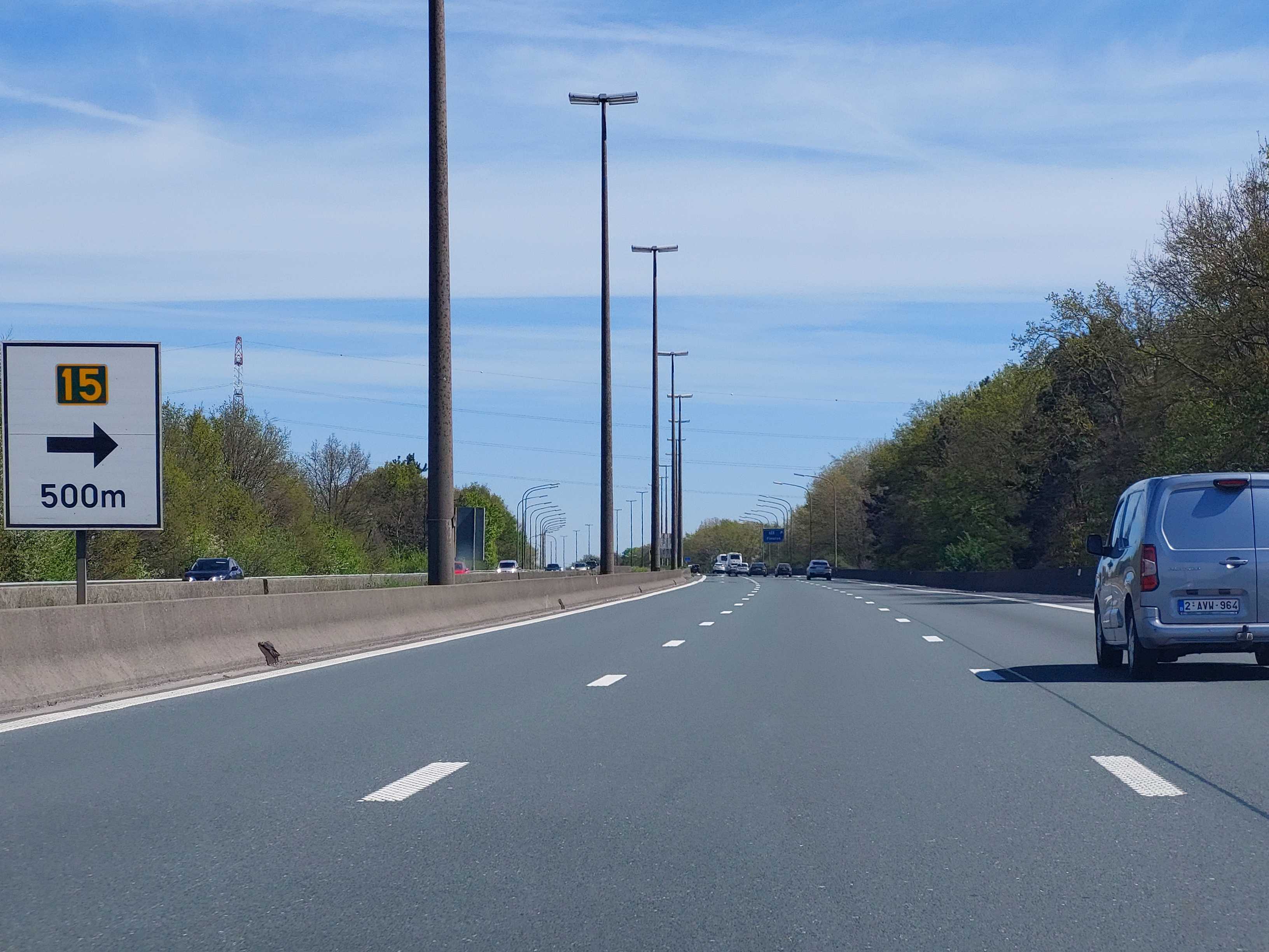 En tant qu'expert en code de la route belge, voici une description détaillée de la situation routière présentée dans l'image, en relation avec la question posée :
Description Détaillée de la Situation Routière
Type de situation routière :
Nous sommes sur une route à chaussées séparées (type autoroute ou voie rapide) comportant trois bandes de circulation dans le sens de notre marche. La chaussée est bien éclairée par des lampadaires alignés. Les accotements sont délimités par des glissières de sécurité en béton sur la gauche (séparant du sens inverse) et un talus végétalisé sur la droite. La visibilité est excellente sous un ciel clair et ensoleillé. La route semble relativement droite dans la portion immédiate, mais présente une légère courbe vers la droite au loin.
Éléments visibles (véhicules, panneaux, feux, marquages au sol, etc.) :
Chaussée et marquages au sol : La chaussée est en bon état. Elle est composée de trois bandes de circulation pour le sens de la photo, séparées par des lignes discontinues blanches. La bande la plus à droite est adjacente à une bande d'arrêt d'urgence, délimitée par une ligne continue blanche. On observe également des lignes vibrantes (marquages rugueux) intégrées aux lignes discontinues séparant les bandes de circulation, destinées à alerter les conducteurs en cas de déport.
Véhicules :
Devant nous : Immédiatement devant le véhicule du conducteur (depuis lequel la photo est prise), sur la bande de circulation de droite, se trouve une camionnette utilitaire légère de couleur grise (plaque d'immatriculation belge "2-AVV-964").
Plus loin : Plusieurs autres véhicules (voitures et une camionnette blanche) sont visibles plus loin sur les différentes bandes de circulation (droite, centrale et gauche), roulant dans le même sens. Le trafic semble fluide.
Panneaux de signalisation : Sur la gauche de la chaussée, un panneau de signalisation vertical est visible :
Il s'agit d'un panneau d'indication, de forme rectangulaire à fond blanc, bordé de noir.
Il comporte un encart jaune avec le chiffre "15" (numéro de sortie/échangeur).
Une flèche noire horizontale pointe vers la droite, indiquant la direction de la sortie.
L'inscription "500m" en noir est placée sous la flèche, indiquant la distance avant cette sortie.
Signification : Ce panneau pré-annonce une sortie d'autoroute ou de voie rapide numérotée 15, qui se situera à 500 mètres sur la droite.
Autres éléments : De nombreux poteaux d'éclairage public sont alignés le long de la chaussée de part et d'autre. Une végétation dense (arbres et buissons) borde la route, créant un corridor végétal. Des lignes électriques aériennes sont visibles au-dessus des arbres sur la gauche.
Position et direction des véhicules :
Le véhicule du conducteur (qui pose la question) se trouve sur la bande de circulation de droite. La camionnette grise que l'on souhaite dépasser est également sur cette même bande, juste devant. Tous les véhicules visibles circulent dans la même direction, vers l'avant.
Panneaux de signalisation présents et leur signification :
Comme décrit ci-dessus, le seul panneau visible est un panneau de pré-signalisation de sortie d'autoroute ou de voie rapide (numéro 15, à 500 mètres sur la droite). Il n'y a pas de panneaux de limitation de vitesse spécifiques ni de panneaux d'interdiction de dépasser visibles dans le champ de l'image.
Tout autre élément pertinent pour comprendre la situation :
La question "Je souhaite dépasser ce véhicule et rester sur cette bande de circulation après le dépassement. Est-ce autorisé ?" est cruciale. Le véhicule à dépasser (la camionnette grise) est sur la bande de droite. Si le conducteur souhaite la dépasser et rester sur la même bande de circulation après le dépassement (c'est-à-dire la bande de droite), cela impliquerait un dépassement par la droite. Si "cette bande de circulation" fait référence à la bande où il se trouverait après avoir dépassé par la gauche* (par exemple, la bande du milieu), la nuance est importante. En l'absence de clarification, l'interprétation la plus directe de "rester sur cette bande" (étant donné que le véhicule du conducteur et celui à dépasser sont sur la bande de droite) suggère un dépassement par la droite.