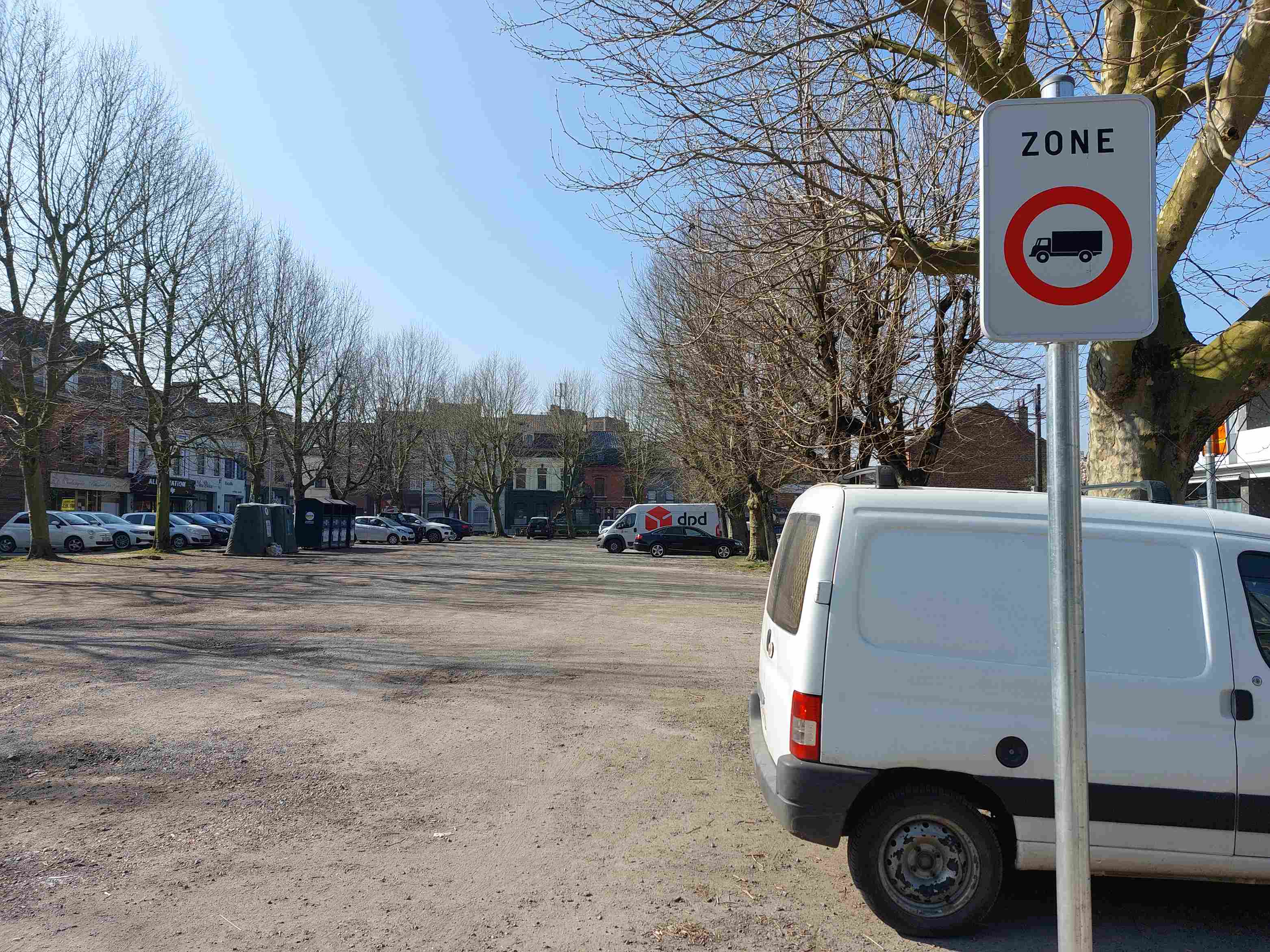 Absolument ! Voici une description détaillée de la situation routière représentée sur l'image, en tant qu'expert en code de la route belge.
Description Détaillée de la Situation Routière pour la Question d'Examen
Type de situation routière :
L'image présente un large espace ouvert, ressemblant à une place publique ou un parking extérieur non asphalté, majoritairement recouvert de graviers ou de terre battue. Ce n'est pas une intersection. La zone est bordée d'arbres à feuilles caduques (dénudés, suggérant l'hiver ou le début du printemps) et de bâtiments en arrière-plan, ce qui indique un environnement urbain ou semi-urbain, probablement le centre d'une localité. L'absence de marquages au sol clairs pour le stationnement sur la surface principale suggère un parking informel ou une aire de stationnement tolérée dans cette zone.
Les éléments visibles :
Ciel : Clair et lumineux, de couleur bleue, indiquant une journée ensoleillée.
Arbres : De nombreux arbres matures, aux branches nues, bordent la place sur les côtés et l'arrière-plan, projetant des ombres.
Sol : La majeure partie de la surface visible est composée de graviers ou de terre battue de couleur claire (brun-grisâtre), avec quelques traces de passages de véhicules.
Bâtiments : En arrière-plan, on distingue une rangée de bâtiments de plusieurs étages, typiques d'une architecture urbaine européenne. Certains semblent abriter des commerces au rez-de-chaussée.
Véhicules :
Au premier plan à droite : L'arrière d'une camionnette blanche de type utilitaire léger (probablement le véhicule du conducteur selon le contexte de la question) est partiellement visible, garée sur le gravier.
Au milieu-plan à gauche : Plusieurs voitures de tourisme sont garées en épi ou en bataille le long d'une limite (possiblement un trottoir invisible ou le bord de la place), face aux bâtiments. On distingue des citadines blanches, un SUV/crossover argenté et une berline sombre.
Au milieu-plan au centre/droite : Plus loin sur la place, une autre camionnette de livraison blanche avec le logo "dpd" est garée, ainsi qu'une berline foncée à côté.
Autres : Quelques autres véhicules sont faiblement visibles plus loin.
Autres structures : Deux grands conteneurs de recyclage (ou de déchets enterrés) de couleur sombre sont visibles derrière les voitures garées à gauche.
Marquages au sol : Aucun marquage au sol de stationnement ou de circulation n'est visible sur la surface en gravier, suggérant que le stationnement est libre dans cette zone délimitée.
La position et direction des véhicules s'il y en a :
Le véhicule au premier plan (celui du conducteur de la question) est une camionnette blanche, garée sur la droite, son arrière tourné vers le centre de l'image.
Les voitures au milieu-plan gauche sont garées en une rangée, face aux bâtiments.
La camionnette "dpd" et la voiture à côté sont garées plus au centre de la place, également stationnaires.
Tous les véhicules visibles sont à l'arrêt, en stationnement.
Les panneaux de signalisation présents et leur signification :
Un panneau de signalisation vertical est clairement visible au premier plan, sur la droite de l'image.
Type de panneau : Il s'agit d'un panneau de début de zone (F107 en Belgique).
Description : Le panneau est carré, à fond blanc, bordé de gris. En haut, le mot "ZONE" est inscrit en lettres majuscules noires. Sous le mot "ZONE", il y a un panneau d'interdiction circulaire classique : un cercle rouge avec une barre oblique (non visible, car le camion n'est pas barré, mais la nature du cercle rouge indique une interdiction) sur fond blanc, et en son centre, la silhouette noire d'un camion (véhicule de transport de marchandises).
Signification : Ce panneau indique le début d'une zone où la circulation de certains véhicules est interdite. Le symbole du camion signifie spécifiquement que les véhicules affectés au transport de choses (marchandises) dont la masse maximale autorisée (MMA) excède 3,5 tonnes sont interdits dans cette zone. L'indication "ZONE" signifie que cette interdiction s'applique à l'ensemble de l'aire délimitée par ce panneau, jusqu'à un panneau de fin de zone (F109) qui annulerait cette restriction.
Tout autre élément pertinent pour comprendre la situation :
Le contexte de la question précise que le conducteur possède une camionnette d'une MMA de 3450 kg (permis B). Cette information est cruciale car la restriction du panneau concerne les véhicules de transport de marchandises dont la MMA dépasse 3,5 tonnes (soit 3500 kg). Le véhicule du conducteur, avec une MMA de 3450 kg, est donc inférieur au seuil d'interdiction de 3500 kg.
Cette description fournit tous les éléments visuels et réglementaires nécessaires pour analyser la question posée.