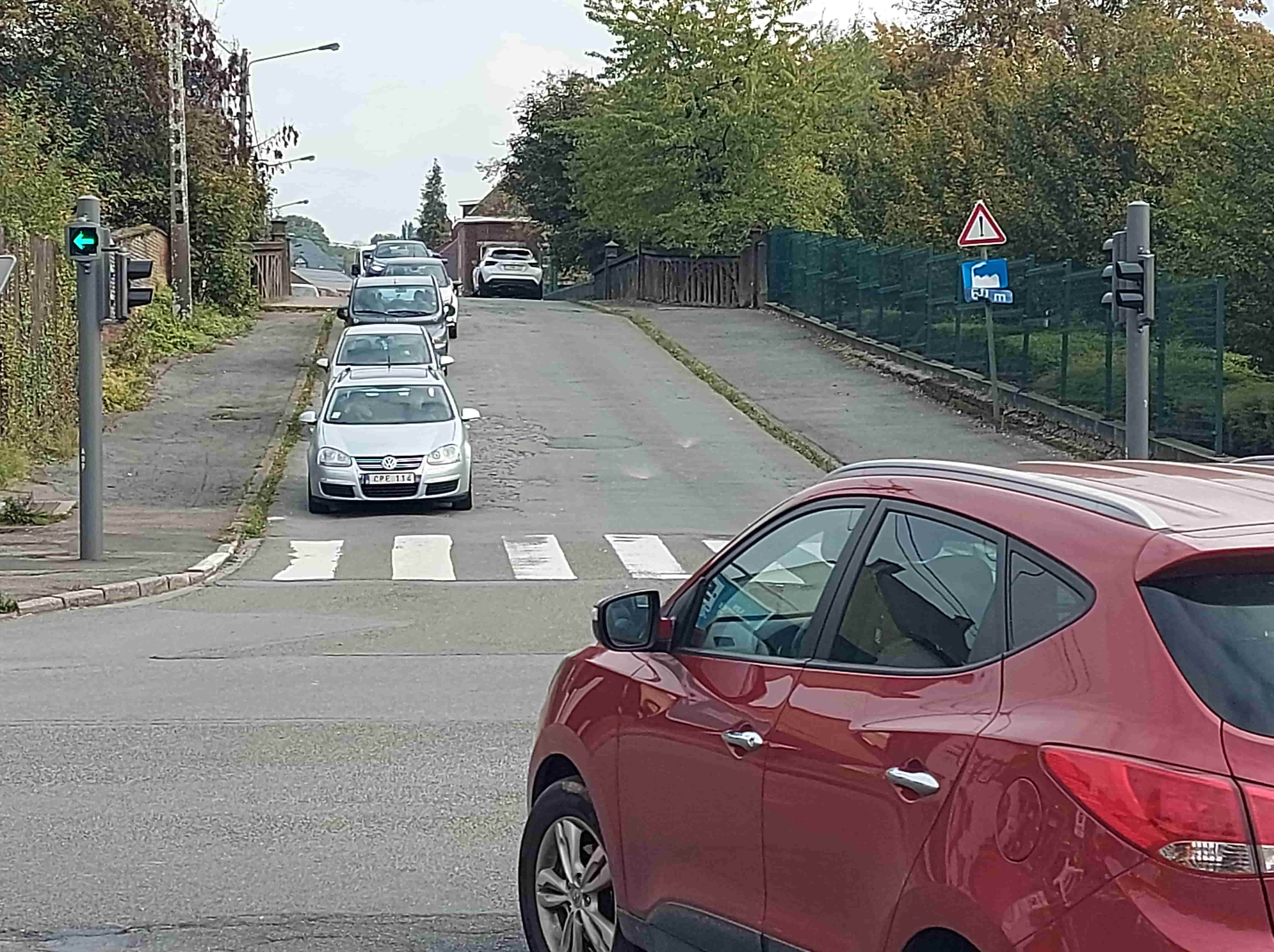 En tant qu'expert du code de la route belge, voici une description détaillée de la situation routière représentée sur l'image, en vue de répondre à la question d'examen.
Description détaillée de la situation routière :
Type de situation routière :
L'image présente un carrefour (intersection) en milieu semi-urbain ou rural. Le conducteur de la voiture rouge se trouve vraisemblablement à l'approche de, ou arrêté à, cet carrefour, qui semble être un croisement entre la route sur laquelle il se trouve et une voie latérale venant de sa gauche. La route principale devant lui semble s'élever légèrement et se prolonge sous une structure (possiblement un pont ou un passage étroit) après une légère courbe. Un passage pour piétons est également présent juste devant la file de véhicules en attente.
Éléments visibles :
Véhicules :
Au premier plan, à droite : Une voiture de type SUV/Crossover de couleur rouge foncé. Seule une partie de l'arrière et du côté droit est visible, suggérant une perspective depuis l'intérieur ou juste derrière ce véhicule. Le conducteur de cette voiture est le sujet de la question.
Devant, sur la route principale : Une file de plusieurs véhicules est arrêtée ou ralentit.
Le véhicule le plus proche de la voiture rouge est une Volkswagen Golf de couleur argentée (immatriculation belge visible : CPE 114), positionnée juste avant le passage pour piétons.
Derrière la Golf, une autre voiture de couleur foncée.
Plus loin dans la file, d'autres véhicules, dont un SUV blanc garé ou arrêté le long du côté droit de la route ascendante.
Feux de signalisation :
Sur un poteau à gauche du carrefour : Un ensemble de feux tricolores.
Un feu principal vertical, dont la couleur n'est pas clairement discernable (potentiellement rouge pour le trafic général traversant l'intersection, ou masqué).
L'élément clé : une flèche verte lumineuse, pointant vers la gauche. Ce feu est situé sur un poteau à gauche de la route principale, et semble réguler le trafic venant d'une voie latérale (à gauche du point de vue de la voiture rouge) et souhaitant tourner à gauche sur la route principale, ou peut-être le trafic sur la route principale qui souhaite bifurquer à gauche. Compte tenu de l'emplacement et de la question, il s'agit d'un feu de "flèche directionnelle" (signal lumineux R2 du Code de la route belge).
Panneaux de signalisation :
À droite de la route principale, au-dessus d'une grille et d'une haie :
Un panneau de danger (A7 : triangle blanc bordé de rouge avec un point d'exclamation noir au centre). Ce panneau signale un danger général qui n'est pas couvert par un panneau de danger plus spécifique.
Juste en dessous, un panneau d'indication rectangulaire bleu (F109 : un pont stylisé au-dessus d'une route avec une hauteur indiquée, difficile à lire précisément mais ressemblant à 2.5m ou 3.5m). Ce panneau indique une limitation de hauteur libre sous un ouvrage d'art (pont, tunnel, etc.).
Marquages au sol :
Juste devant la Volkswagen Golf : Un passage pour piétons, matérialisé par des bandes blanches longitudinales (zébras).
Autres éléments :
Végétation : De nombreux arbres et buissons bordent la route des deux côtés, donnant un aspect verdoyant à l'environnement.
Infrastructures : Des trottoirs ou des accotements sont visibles, avec des bordures. Des poteaux (électriques ou de signalisation) sont implantés. Une clôture grillagée verte et une palissade en bois sont visibles à droite. Un petit bâtiment en briques ou en parpaings est visible en retrait sur la droite, après la file de voitures.
Position et direction des véhicules :
La voiture rouge : Est positionnée sur une voie, potentiellement une voie d'accès au carrefour ou la voie principale. Son orientation suggère qu'elle s'apprête à continuer tout droit ou à tourner à droite sur la route principale.
La file de voitures : Ces véhicules sont alignés sur la route principale, apparemment arrêtés au niveau du passage pour piétons, dans la même direction que la voiture rouge envisage de prendre. Ils semblent attendre une opportunité de passage ou un changement de feu.
Le feu avec la flèche verte est destiné au trafic venant d'une voie située à gauche de la route principale où se trouve la voiture rouge, et qui souhaite s'engager vers la gauche dans l'intersection.
Signification des panneaux de signalisation présents :
Panneau A7 (Danger général) : Avertit les usagers de la route d'un danger non spécifié, nécessitant une vigilance accrue et une adaptation de la conduite. Ce danger pourrait être lié à la configuration complexe du carrefour, à la présence d'une structure (pont) ou à l'état de la chaussée.
Panneau F109 (Hauteur libre limitée à ~3.5m) : Indique la hauteur maximale autorisée pour les véhicules sous la structure à venir (probablement le pont visible plus loin sur la route ascendante). Les véhicules dont la hauteur excède cette limite doivent emprunter un autre itinéraire.
Tout autre élément pertinent pour comprendre la situation :
La question se concentre sur la flèche verte à gauche. En Belgique, une flèche verte sur un feu tricolore signifie que le conducteur est autorisé à s'engager dans la direction indiquée par la flèche (ici, à gauche), mais il doit céder le passage aux usagers de la route (y compris les piétons) qui circulent sur la ou les autres branches du carrefour et qui bénéficient du droit de passage. S'il s'agit d'une flèche verte fixe (non clignotante) accompagnant un feu vert plein pour la voie, alors elle confirme la possibilité de tourner dans cette direction sans condition de céder le passage si l'intersection est protégée. Cependant, l'usage le plus courant d'une flèche verte séparée est d'autoriser un mouvement sous condition de céder le passage.
La question indique ensuite que "Le conducteur de la voiture rouge a la priorité de passage par rapport :". Cela sous-entend que la voiture rouge se trouve sur une voie où elle a la priorité, et la flèche verte est pour un mouvement potentiellement conflictuel venant d'une autre branche du carrefour. Il est probable que la voiture rouge soit sur la voie principale et bénéficie d'un feu vert (non visible sur l'image pour sa direction spécifique) ou de la priorité de droite (ce qui serait peu probable avec des feux de signalisation).