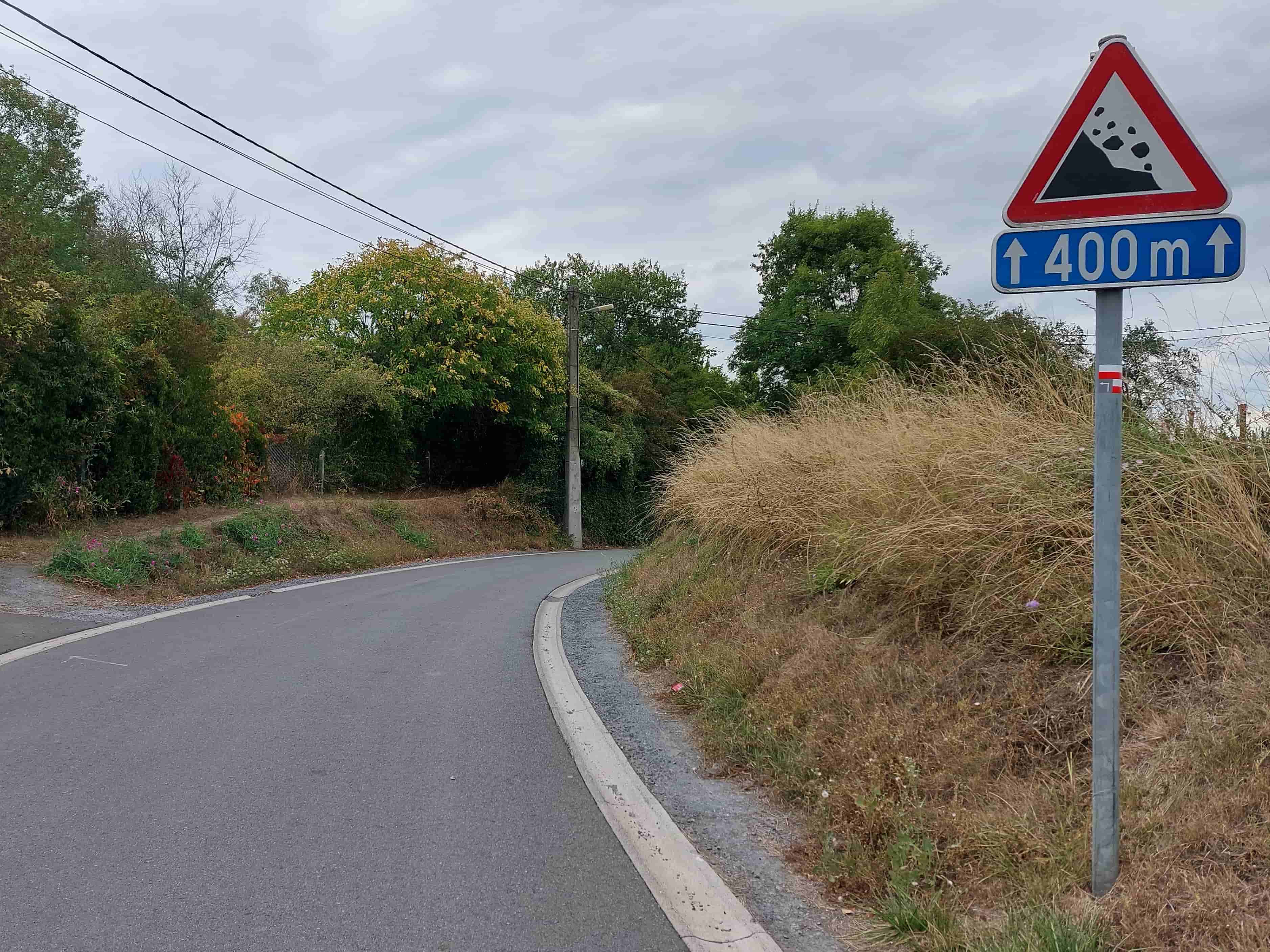 En tant qu'expert en code de la route belge, voici une analyse détaillée de l'image pour comprendre le contexte de la question :
Description détaillée de la situation routière :
Type de situation routière :
La scène représente une route asphaltée, vraisemblablement à double sens de circulation (bien qu'aucune ligne centrale ne soit visible, c'est l'hypothèse la plus commune pour ce type de chaussée sans indication contraire), qui décrit une courbe prononcée vers la gauche. Le côté droit de la chaussée est bordé par un accotement herbeux en pente douce, puis un talus plus abrupt couvert de végétation sèche et d'arbres. Le côté gauche de la route est également bordé d'une végétation dense. Il s'agit d'un environnement qui semble plus proche d'une zone rurale ou péri-urbaine que d'un centre-ville dense, mais compatible avec les périphéries ou zones vertes de Bruxelles. Il n'y a pas d'intersection, de carrefour giratoire ou de feu de signalisation visible.
Éléments visibles :
Chaussée : Asphalte gris foncé, en bon état apparent, décrivant une courbe vers la gauche. Une ligne continue blanche délimite le bord droit de la chaussée. Un caniveau/bordure en béton gris clair sépare la chaussée de l'accotement sur le côté droit.
Accotements et végétation : À droite, un accotement couvert d'herbes sèches et hautes s'élève en un talus boisé. À gauche, la route est également bordée par une dense végétation (arbres et buissons). L'ensemble de la végétation suggère une fin d'été ou un début d'automne.
Infrastructure : Un poteau en béton (probablement électrique ou de télécommunication) est visible dans la courbe, sur le côté gauche de la route, avec des câbles aériens courant au-dessus de la chaussée. Un poteau métallique gris supporte les panneaux de signalisation sur le côté droit de la route.
Véhicules : Aucun véhicule n'est visible sur la route ou ses abords. La perspective est celle d'un conducteur circulant sur cette route.
Feux : Aucun feu de signalisation n'est visible.
Marquages au sol : Seule une ligne continue blanche délimitant le bord droit de la chaussée est visible.
Position et direction des véhicules s'il y en a :
Il n'y a pas de véhicules visibles dans l'image. Le point de vue est celui du conducteur qui aborde la courbe et se dirige vers les panneaux de signalisation.
Panneaux de signalisation présents et leur signification (Code de la Route Belge) :
Les panneaux sont montés sur un seul poteau, sur le côté droit de la route :
Panneau supérieur : Il s'agit d'un panneau de danger triangulaire avec un liseré rouge, un fond blanc et un symbole noir. Le symbole représente une montagne ou une pente d'où tombent des rochers sur la chaussée.
Identification : C'est le panneau de danger A9 - Chutes de pierres ou présence de pierres sur la chaussée.
Signification : Ce panneau avertit les usagers de la route qu'ils approchent d'un tronçon de route où il existe un risque de chutes de pierres (venant du talus à droite dans ce cas précis) ou de présence de pierres déjà tombées sur la chaussée. Les conducteurs doivent faire preuve de prudence, adapter leur vitesse et être prêts à réagir.
Panneau inférieur (panonceau) : Il s'agit d'un panonceau rectangulaire à fond bleu avec des symboles et du texte blancs. On y voit deux flèches verticales pointant vers le haut, encadrant l'indication "400 m".
Identification : C'est un panonceau de distance (type P1), spécifiquement un panonceau de longueur ou d'étendue.
Signification : Les flèches pointant vers le haut de part et d'autre de la distance indiquent que le danger signalé par le panneau A9 (chutes de pierres) s'étend sur une longueur de 400 mètres à partir de l'emplacement du panneau. Il ne signifie pas que le danger est à 400 mètres, mais qu'il commence ici et se poursuit sur 400 mètres.
Tout autre élément pertinent pour comprendre la situation :
Marquage de sentier : Sur le poteau métallique, juste en dessous des panneaux de signalisation, on peut observer un petit marquage rectangulaire (blanc avec une bande rouge horizontale au-dessus) et un symbole en forme de "L" ou de flèche pointant vers la gauche et vers le bas. Il s'agit très probablement d'un balisage de sentier de randonnée (souvent GR - Grande Randonnée en Belgique), indiquant une direction ou un point de repère pour les marcheurs, et n'a pas de signification pour la circulation routière.
Ciel : Le ciel est couvert et gris, indiquant une luminosité diffuse, sans ensoleillement direct.
• Contexte "Bruxelles" : Bien que l'image évoque un cadre moins urbain, la Région de Bruxelles-Capitale inclut des zones vertes, des parcs et des routes périphériques où ce type de signalisation et d'environnement est tout à fait plausible.
En résumé, la question porte sur la signification combinée du panneau de danger "chutes de pierres" et du panonceau de "longueur" indiquant que ce danger s'étend sur 400 mètres à partir de ce point sur la route.