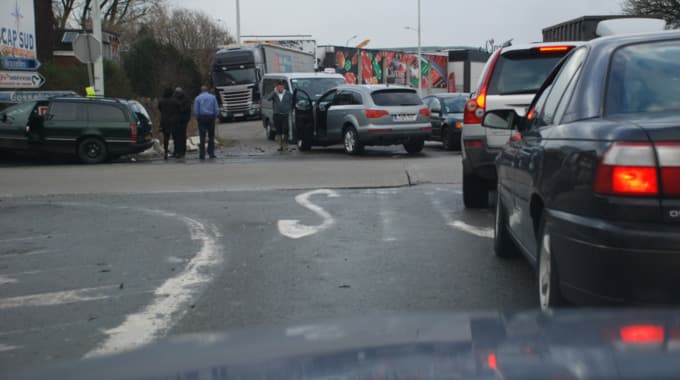 Voici une description détaillée de l'image, analysée comme un expert en code de la route belge, pour comprendre le contexte de la question d'examen.
Description détaillée de la situation routière
L'image présente une scène routière complexe et potentiellement perturbée, prise depuis l'intérieur d'un véhicule à l'arrêt, probablement en file. Le temps est couvert, et la chaussée semble mouillée, suggérant une pluie récente ou un temps humide.
Type de situation routière:
Il s'agit d'une intersection ou d'un carrefour important, situé dans un environnement urbain ou péri-urbain, vraisemblablement à l'entrée ou la sortie d'une zone commerciale ou industrielle. La présence de nombreux véhicules et de panneaux de direction confirme qu'il s'agit d'un point de jonction où plusieurs voies se rencontrent ou se séparent. Une partie de la scène, sur la gauche, semble indiquer un événement (accident ou arrêt prolongé) ayant potentiellement attiré l'attention.
Éléments visibles:
Véhicules en file (premier plan et milieu droit):
Le véhicule du photographe est arrêté, son tableau de bord étant partiellement visible en bas de l'image.
Juste devant, une voiture de tourisme noire (ressemblant à une Opel Omega B Caravan) est également à l'arrêt, ses feux stop rouges étant allumés.
Devant la voiture noire, un SUV gris clair (ressemblant à un Volvo XC90) est aussi à l'arrêt, ses feux stop allumés.
Plusieurs autres véhicules sont visibles plus loin dans la file ou dans le sens opposé.
Véhicules à l'arrêt sur le côté gauche (potentiellement impliqués ou stationnés):
Une grande voiture break de couleur vert foncé (ressemblant à une Opel Omega A Caravan) est stationnée ou arrêtée en bord de chaussée, sur la gauche. La portière conducteur est ouverte.
À côté, un SUV gris clair (ressemblant à une Audi Q7) est également arrêté, la portière conducteur ouverte.
Derrière l'Audi Q7, on distingue une camionnette ou un utilitaire de couleur claire, également à l'arrêt.
Des poids lourds (camions, dont un Scania reconnaissable) sont visibles en arrière-plan, suggérant la proximité d'une zone d'activité ou de logistique.
Personnes:
Un groupe de trois à quatre personnes se tient debout près du break vert foncé, au bord de la chaussée. Leurs postures suggèrent qu'elles discutent ou évaluent une situation. L'une d'elles, en chemise bleue, semble regarder vers le sol ou vers le groupe.
Une autre personne est visible près du SUV Audi Q7.
Panneaux de signalisation:
Sur le côté gauche, un grand panneau de signalisation de direction (à fond blanc avec des flèches, indiquant des destinations) est clairement visible. On peut lire "CAP SUD", "Bruxelles" et "Gosselies".
Sous ce panneau de direction, un panneau d'arrêt obligatoire (STOP) est présent. Il s'agit du panneau B5 du code de la route belge, de forme octogonale et de couleur rouge avec l'inscription "STOP" en blanc.
Sous le panneau STOP, un petit panneau additionnel (panonceau) est visible, mais illisible.
Marquages au sol:
Une grande flèche blanche courbée vers la gauche est peinte sur la chaussée au premier plan, indiquant une direction obligatoire ou conseillée (un virage à gauche ou une voie de tourner à gauche).
Des lignes blanches continues ou discontinues sont également visibles, délimitant la chaussée.
Autres éléments:
Des arbres et buissons au feuillage clairsemé (hiver ou début de printemps).
Des bâtiments industriels ou commerciaux en arrière-plan avec des affiches publicitaires.
Position et direction des véhicules:
Les véhicules au premier plan et milieu droit (celui du photographe, la berline noire, le SUV gris) sont arrêtés en file, orientés vers l'avant, attendant de pouvoir avancer ou s'engager dans l'intersection.
Les véhicules sur le côté gauche (break vert, SUV Audi Q7, camionnette) sont stationnés ou arrêtés sur le bas-côté, légèrement en retrait de la circulation principale. Le break vert est parallèle à la chaussée, tandis que l'Audi Q7 est légèrement inclinée. Les portières ouvertes indiquent une intervention ou une activité en cours.
Panneaux de signalisation présents et leur signification:
Panneau de direction (blanc avec flèches): Il informe les conducteurs des différentes directions et destinations accessibles depuis cette intersection, comme "Bruxelles" et "Gosselies", permettant de planifier leur itinéraire.
• Panneau STOP (B5): Ce panneau impose à tout conducteur de marquer un arrêt complet à la ligne d'arrêt (ou à l'aplomb du signal si pas de ligne) et de céder le passage à tous les usagers circulant sur la ou les chaussées qu'il va aborder. Sa présence confirme que le carrefour est une intersection à priorité où le conducteur venant de la voie où se trouve le panneau doit obligatoirement s'arrêter.
Tout autre élément pertinent pour comprendre la situation:
La présence d'un groupe de personnes rassemblées autour de véhicules dont les portières sont ouvertes, à proximité d'une intersection, et le contexte d'une question d'examen sur les premiers secours ("Un blessé a une grande coupure au bras qui saigne beaucoup. Peut-on mettre un garot ?") suggèrent fortement qu'un incident (potentiellement un accident de la circulation ou une situation d'urgence) a eu lieu à cet endroit ou à proximité immédiate. L'image dépeint donc la scène d'un événement qui a requis l'arrêt des véhicules et l'attention des personnes présentes, créant un contexte pour une question de premiers secours. Le conducteur du véhicule d'où est prise la photo est témoin de cette situation.