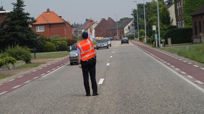 En tant qu'expert en code de la route belge, voici une description détaillée de la situation présentée dans l'image, en relation avec la question posée :
Description détaillée de la situation routière :
Type de situation routière :
La scène se déroule sur une route asphaltée bidirectionnelle, traversant ce qui semble être une zone semi-urbaine ou résidentielle de village/périphérie. Il n'y a pas d'intersection majeure visible immédiatement devant l'agent, mais la présence de plusieurs véhicules suggère un flux de circulation habituel. La situation est caractérisée par une intervention d'un agent de la force publique pour réguler le trafic.
Éléments visibles :
Agent qualifié : Au centre de l'image, un agent de police (reconnaissable à sa casquette et son gilet fluo orange portant l'inscription "POLITIE") est debout au milieu de la chaussée. Il est positionné de dos par rapport à la caméra, faisant face au sens de circulation des véhicules venant de loin.
Geste de l'agent : Son bras droit est levé verticalement, le poing serré. Ce geste est une injonction claire de l'agent.
Chaussée : La route est asphaltée et semble comporter deux voies de circulation (une par sens). La surface est sèche.
Marquages au sol :
Des lignes blanches discontinues sont peintes au centre de la chaussée, délimitant les deux sens de circulation.
Une ligne blanche continue sépare la chaussée principale d'une bande rouge sur le côté droit (du point de vue de la caméra).
La bande rouge est elle-même bordée d'une ligne blanche discontinue sur son côté extérieur.
Piste cyclable/Bande cyclable suggérée : La bande rouge sur le côté droit de la route est très probablement une piste cyclable ou une bande cyclable, caractéristique des infrastructures routières belges pour les vélos.
Véhicules :
Plusieurs véhicules circulent ou sont à l'arrêt devant l'agent.
Un véhicule gris clair est visible assez proche de l'agent, dans la voie de circulation qui se dirige vers lui. Il semble en train de s'arrêter ou est déjà immobilisé.
Un autre véhicule est visible derrière le véhicule gris clair.
Plus loin sur la route, d'autres véhicules sont visibles, indiquant un flux de trafic.
Sur le côté gauche de la route (par rapport à la caméra), un véhicule gris clair est également arrêté, le long de la "piste cyclable" (du point de vue de la caméra) ou sur le bord de la route.
Signalisation verticale :
Un panneau d'arrêt de bus (panneau bleu rectangulaire avec un pictogramme de bus blanc) est visible sur le côté droit de la route, après la piste cyclable, indiquant un emplacement d'arrêt pour les transports en commun.
Des lampadaires sont présents des deux côtés de la route.
Environnement : Des maisons individuelles en briques rouges avec des toits en tuiles sont visibles des deux côtés de la route, entourées de verdure (arbres, buissons, pelouses). Le ciel est clair, suggérant une journée ensoleillée.
Position et direction des véhicules s'il y en a :
L'agent est positionné de manière à intercepter le trafic venant de la direction qu'il regarde. Les véhicules devant lui (le gris clair et celui derrière) sont en approche ou déjà à l'arrêt, faisant face à l'agent. Le véhicule sur le côté gauche (devant le bosquet) est également immobilisé, suggérant qu'il attend l'instruction ou qu'il a déjà été arrêté. Tous les véhicules concernés par le geste de l'agent doivent s'immobiliser.
Panneaux de signalisation présents et leur signification :
Le seul panneau de signalisation clairement identifiable est le panneau d'arrêt de bus, qui indique un arrêt pour les véhicules de transport en commun. Il n'y a pas d'autres panneaux de signalisation routière régulatoire (comme "STOP", "Cédez le passage", limitation de vitesse) clairement visibles et directement pertinents pour l'injonction de l'agent. La primauté revient ici au geste de l'agent.
Tout autre élément pertinent pour comprendre la situation :
Le point le plus pertinent est l'injonction de l'agent. Selon l'article 69.1.1° de l'Arrêté Royal du 1er décembre 1975 portant règlement général sur la police de la circulation routière et de l'usage de la voie publique (Code de la route belge) :
"L'agent qui lève un bras verticalement, le poing fermé, ordonne l'arrêt à tous les usagers de la route qui, quelle que soit leur direction, s'approchent de lui. Si cet ordre est donné à une intersection, les usagers qui se trouvent déjà dans l'intersection ou très près de celle-ci peuvent la dégager."
Dans ce contexte, le geste de l'agent signifie une obligation formelle pour tous* les usagers de la route qui s'approchent de lui (provenant de la direction vers laquelle il fait face, mais aussi potentiellement des autres directions non visibles directement de la caméra, ou des voies latérales) de s'arrêter immédiatement. L'agent exerce son autorité pour réguler le trafic, probablement en raison d'un événement (accident, contrôle, travaux, passage spécial, etc.) non visible sur l'image.