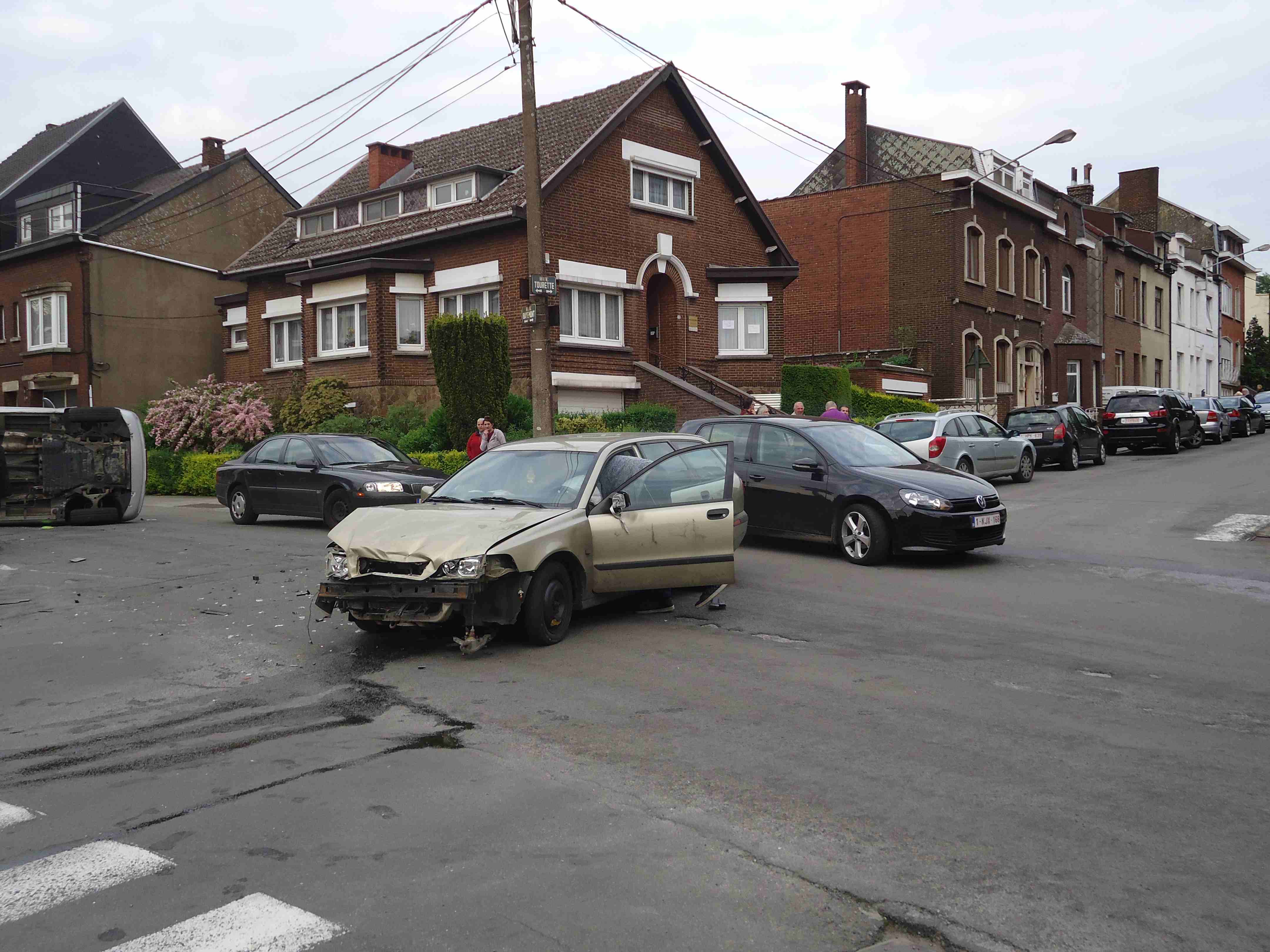En tant qu'expert en code de la route belge, voici une description détaillée de la situation routière présentée sur l'image, pertinente pour comprendre le contexte de la question d'examen :
Description Détaillée de la Situation Routière (Accident):
Type de situation routière:
L'image montre la scène d'un accident grave survenu à une intersection (carrefour) dans une zone résidentielle ou urbaine.
Il s'agit d'une rue locale, bordée de maisons en briques, avec des trottoirs et un éclairage public.
Un passage pour piétons (bandes blanches ou "zébra") est clairement visible en premier plan, traversant la chaussée, indiquant une zone où les piétons ont la priorité.
Éléments visibles (véhicules, panneaux, feux, marquages au sol, etc.):
Véhicules impliqués dans l'accident:
Une voiture de couleur beige clair/doré (vraisemblablement une Volvo V40 ou S40 ancienne génération) est positionnée au centre de l'intersection. Elle présente des dommages frontaux très importants : le pare-chocs avant est arraché, les phares sont cassés et le capot est déformé. La porte du conducteur est ouverte. C'est le véhicule du chauffeur mentionné dans la question.
Un véhicule de couleur gris argenté (une petite berline ou compacte, par exemple une Renault Clio) est renversé sur son flanc gauche, reposant sur le côté près du trottoir à gauche de la voiture beige. Il semble également très endommagé.
Autres véhicules:
Une voiture noire (une Volkswagen Golf de génération récente, immatriculée 1-KJX-168) est arrêtée ou stationnée sur la droite de la voiture beige, face à l'observateur. Elle ne semble pas endommagée et pourrait être un témoin ou un véhicule arrivé après l'accident.
Plusieurs autres voitures sont stationnées le long du trottoir sur la droite de la rue (une Citroën C3 noire, un SUV noir, plusieurs berlines grises ou argentées). Une berline gris foncé est également garée sur le côté gauche de la rue, juste après le véhicule renversé.
Personnes: Plusieurs personnes sont visibles, observant la scène, témoignant de l'ampleur de l'événement. Certaines sont proches des véhicules accidentés, d'autres sont en arrière-plan.
Panneaux de signalisation:
Sur le poteau téléphonique/électrique à droite de la voiture beige, un panneau de nom de rue indique clairement "RUE TOURETTE".
Juste en dessous, un autre panneau de nom de rue est partiellement visible, indiquant probablement une autre rue (ex: "RUE DU HAUT PONT").
Important : Aucun panneau de priorité (Cédez le passage B1, Stop B5) n'est visible pour l'intersection immédiate où l'accident s'est produit.
Feux de signalisation: Aucun feu de signalisation n'est visible à l'intersection.
Marquages au sol:
Un passage pour piétons (zébra) est clairement visible en premier plan, traversant la chaussée.
Des traces de freinage (ou de ripage) et des débris (fragments de plastique, de verre, pièces de carrosserie) sont éparpillés sur la chaussée autour des véhicules accidentés.
Des flaques de liquide (probablement huile, liquide de refroidissement) sont visibles sur l'asphalte près de la voiture beige.
Environnement: Des maisons en briques avec des toits en pente, des buissons et des arbres (dont un avec des fleurs roses) bordent les rues. Le ciel est couvert.
Position et direction des véhicules s'il y en a:
La voiture beige clair est positionnée de manière transversale sur la chaussée, le capot fortement endommagé étant orienté approximativement vers la caméra et légèrement vers la gauche. Elle semble avoir subi un impact latéral important ou avoir pivoté après la collision.
La voiture grise/argentée est renversée sur son flanc gauche, sa partie arrière étant orientée vers l'observateur et sa partie avant vers l'intérieur de l'image (vers la droite par rapport à sa direction initiale). Elle se trouve à gauche et légèrement en avant de la voiture beige.
La voiture noire (VW Golf) est arrêtée sur la droite, face à l'observateur, son avant étant aligné avec la direction de la rue principale.
Panneaux de signalisation présents et leur signification:
Les panneaux "RUE TOURETTE" et le panneau partiellement visible en dessous servent uniquement à identifier les noms des rues.
L'absence notable de panneaux de priorité (B1 ou B5) est cruciale. En l'absence de signalisation spécifique à une intersection en Belgique, la règle générale de la priorité de droite s'applique.
Tout autre élément pertinent pour comprendre la situation:
L'état des véhicules et les débris sur la chaussée indiquent une collision d'une certaine violence.
Le fait que l'accident se déroule à un carrefour sans signalisation de priorité explicite implique que les conducteurs devaient appliquer la règle de la priorité de droite.
La question se concentre sur le conducteur de la voiture beige clair, dont l'état d'ébriété (0,55 Mgr/l) est le facteur aggravant principal pour la décision du Procureur du Roi concernant le retrait de permis. L'image fournit le contexte visuel de cet accident.