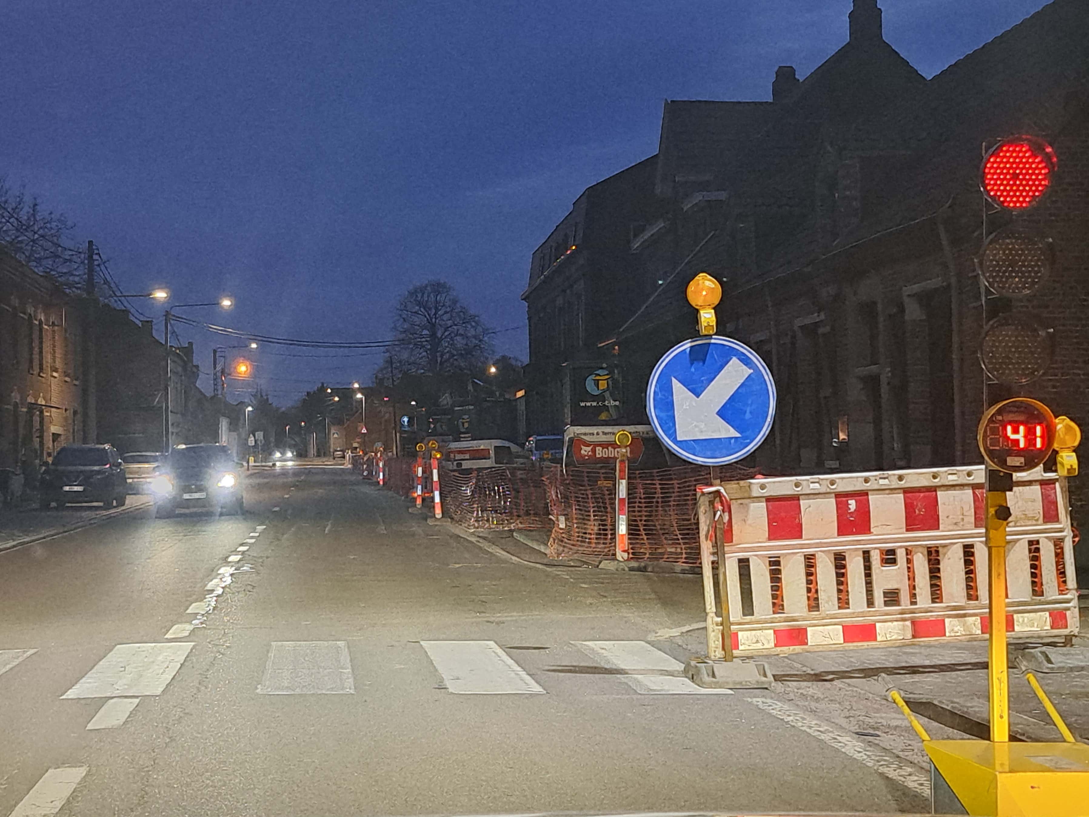 En tant qu'expert en code de la route belge, voici une description détaillée de la situation présentée dans l'image :
Description détaillée de la situation routière :
Type de situation routière :
La scène se déroule sur une route urbaine ou de village, à la tombée de la nuit ou au petit matin (luminosité crépusculaire). Il s'agit d'une zone affectée par d'importants travaux routiers (chantier), ce qui a provoqué un rétrécissement de la chaussée et la mise en place d'une signalisation temporaire pour gérer le trafic. La circulation est gérée de manière alternée par des feux de chantier synchronisés. Le véhicule d'où est prise la photo est à l'arrêt, face à un passage pour piétons et un feu de circulation.
Éléments visibles :
Chaussée et marquages au sol :
Une chaussée asphaltée est visible.
Juste devant le véhicule qui prend la photo, on distingue clairement un passage pour piétons (marquage blanc de rectangles parallèles).
Aucune ligne continue ou discontinue n'est visible pour délimiter les voies de circulation classiques, probablement en raison des travaux qui ont modifié la configuration normale de la route.
Véhicules :
Le véhicule du conducteur (implicite) est à l'arrêt.
Plusieurs véhicules sont visibles dans la voie opposée, s'approchant en direction du véhicule du conducteur. Leurs phares sont allumés, confirmant la faible luminosité ambiante. On distingue au moins deux voitures distinctes, la première étant un SUV sombre, suivi d'une autre voiture.
Panneaux de signalisation :
Panneau D1a (obligation de contourner par la droite) : Directement à droite de la voie de circulation, un panneau circulaire bleu avec une flèche blanche oblique pointant vers le bas à droite est clairement visible. Ce panneau signifie "Sens obligatoire de franchissement : contournement de l'obstacle par la droite".
Feu jaune clignotant : Au-dessus du panneau D1a, une balise lumineuse orange (feu jaune clignotant) est activée. Elle indique qu'il s'agit d'une signalisation temporaire liée à un danger (les travaux).
Un autre panneau de danger triangulaire est très faiblement visible plus loin sur la gauche, mais illisible.
Feux de signalisation :
Feu de chantier temporaire rouge : À l'extrême droite de l'image, un feu de signalisation vertical est présent, montrant un feu rouge allumé.
Compteur numérique : Sous le feu rouge, un petit écran digital affiche le nombre "41", indiquant probablement qu'il reste 41 secondes avant que le feu ne change de couleur. Ce sont des feux de chantier gérant un passage alterné.
Une balise lumineuse orange (feu jaune clignotant) est également présente sous le décompte du feu de chantier, pour renforcer la signalisation temporaire.
Signalisation et aménagements de chantier :
De nombreuses barrières de chantier rouges et blanches sont alignées le long du côté droit de la route, délimitant la zone de travaux.
Des filets de sécurité orange (grillages de chantier) sont également visibles derrière les barrières, indiquant une zone de fouilles ou un obstacle à ne pas franchir.
Plus loin dans le chantier, on aperçoit du matériel de chantier (par exemple, un logo "Bobcat").
Environnement général :
Des bâtiments (maisons ou petits immeubles) bordent la route des deux côtés, typiques d'une agglomération.
Des lampadaires sont allumés, éclairant la chaussée.
Des arbres dénudés sont visibles en arrière-plan, ainsi qu'un ciel sombre (nuit ou crépuscule).
Des câbles électriques ou de télécommunication traversent la scène en hauteur.
Position et direction des véhicules :
Le véhicule du conducteur est à l'arrêt, positionné devant le passage pour piétons et le feu rouge de chantier.
Les véhicules d'en face circulent en direction opposée au véhicule du conducteur. Ils sont en mouvement, leurs phares illuminant la scène.
Panneaux de signalisation et leur signification :
Panneau D1a (rond bleu, flèche blanche oblique bas-droite) : C'est un signal de prescription (obligation). Il indique que les conducteurs doivent contourner l'obstacle (les travaux) par la droite.
Feu jaune clignotant (sur le panneau et près du feu de chantier) : Indique une situation temporaire, un danger ou des travaux.
Feu rouge de chantier : Signifie "STOP" absolu. Les véhicules doivent s'arrêter avant le passage pour piétons et attendre que le feu passe au vert. Le compteur "41" indique le temps d'attente restant en secondes.
Tout autre élément pertinent :
La présence combinée des travaux, du panneau d'obligation de contournement, du feu rouge temporaire avec décompte, et du passage pour piétons juste devant le véhicule, crée une situation qui exige une attention particulière et le respect strict de la signalisation de chantier et des feux de circulation. La faible luminosité accentue le besoin de vigilance. Le système de feux de chantier avec décompte gère le passage alterné sur une voie unique.
Contexte de la question : "Dans cette situation, je dois utiliser :"
La question vise probablement à évaluer les connaissances du conducteur concernant :
L'arrêt obligatoire au feu rouge.
La prise en compte du décompte.
L'attitude à adopter face à un passage pour piétons.
L'utilisation appropriée des feux du véhicule (par exemple, les feux de croisement, ou l'absence de feux de route si des véhicules arrivent en face ou si l'éclairage est suffisant).
L'anticipation du contournement de l'obstacle par la droite une fois le feu vert.